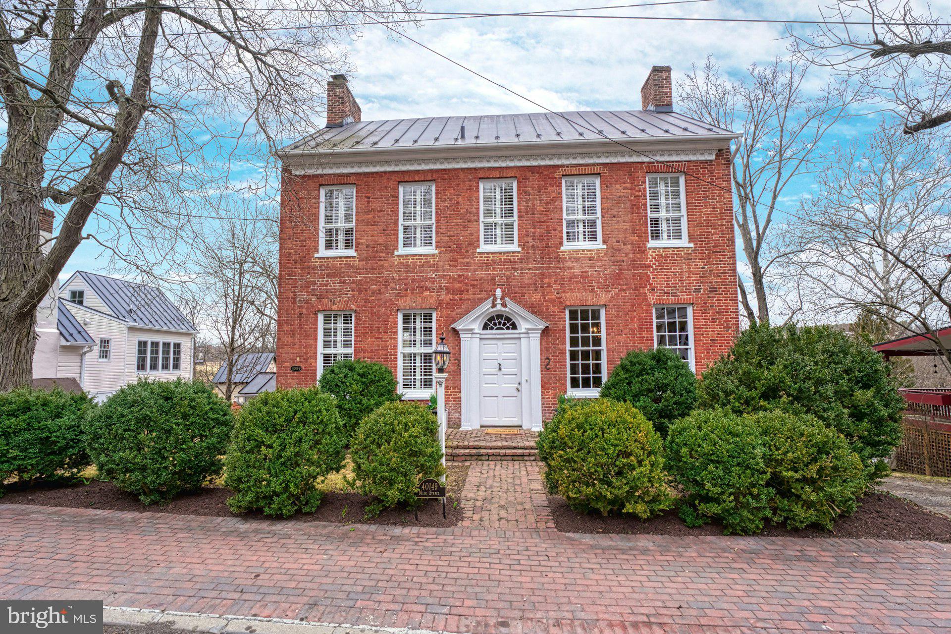 The "Bank House" – Circa 1806
Located in the heart of historic Waterford, "The Bank House" is a stunning example of Federal architecture dating back to 1806. Carefully restored over the years, this home retains its original woodworking, hardware, and wide-plank floors while seamlessly incorporating modern comforts.
Stepping into the grand foyer, you'll be welcomed by high ceilings and a beautifully crafted staircase. The modern eat-in kitchen, designed for both style and practicality, opens to a mudroom and back porch, creating a bright and airy living space. The large dining room, complete with a wood-burning fireplace, flows into the elegant paneled library with wood stove is the heart of the home.
Upstairs, you'll find five spacious bedrooms and two full baths, offering ample room for family and guests. A full walk-up attic provides generous storage space.
The beautifully landscaped lot backs to 144 acres of preserved open land with scenic walking trails and a tranquil creek. Enjoy outdoor living on the large side porch or dine al fresco on the stone patio. The property also includes the original spring house and a charming smokehouse, now serving as a garden shed. Located in western Loudoun, this historic treasure offers a peaceful retreat while remaining convenient to local wineries, breweries, and Leesburg. Dulles Airport is just 25 minutes away, and Washington, D.C., is only 30 miles from your doorstep.

A rare opportunity to own a piece of history in the quaint village of Water