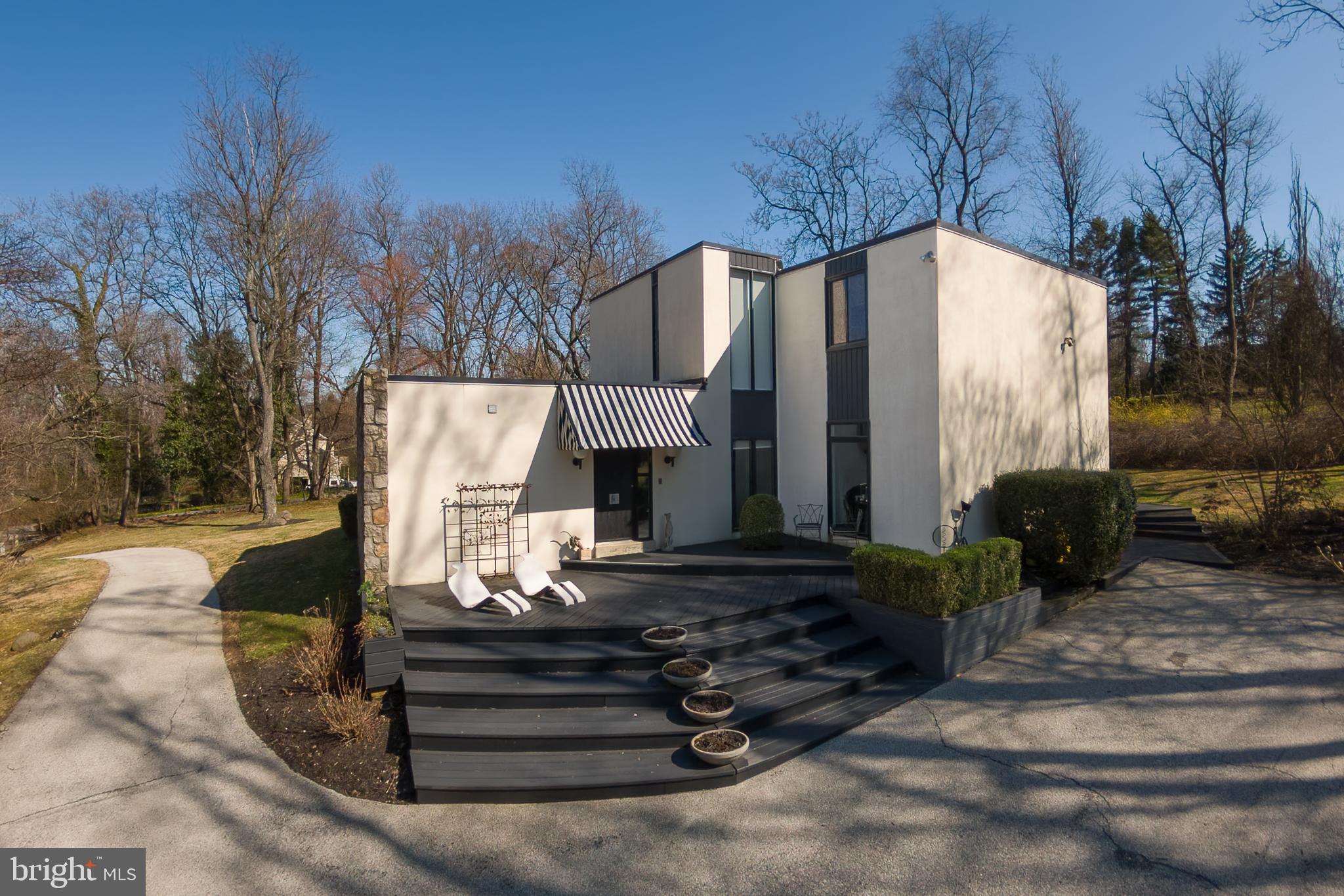 First time on the market since its custom construction in 1973, this striking four-bedroom, three-and-a-half-bath contemporary residence is sited on a beautifully elevated 1.07-acre parcel in Villanova’s coveted Lower Merion Township, tucked along a quiet double cul-de-sac.
Wide, terraced steps lead to a handsome foyer where lacquered black hardwood floors immediately set the tone. Owned and designed by a professional interior designer, the home is a confident study in black and white—highly styled, yet grounded by an abundance of oversized windows that flood the interiors with natural light and draw the surrounding landscape inward.

To the left of the foyer, a sunken den offers a welcoming retreat, complete with custom shelving and an en-suite bath—ideal for intimate conversation, a private office, or gracious guest accommodations. To the right, the expansive family room features 9½-foot ceilings, custom windows, and sliding doors that open to the rear terrace, creating an easy flow between indoor and outdoor living. This space connects seamlessly to the breakfast room and kitchen beyond.
The kitchen is both sophisticated and highly functional, anchored by a black granite island with cooktop, double Dacor wall ovens, stainless steel refrigeration, and thoughtfully designed cabinetry. Generous storage throughout the kitchen and breakfast area ensures everyday convenience with everything close at hand.

Through a wide cased opening, the dramatic dining room offers flexibility for two distinct seating areas. Long horizontal windows and a stylized feature wall add architectural interest and depth. Just beyond, a more formal living space is centered around a wood-burning fireplace, creating a warm and elegant gathering place. A beautifully appointed powder room completes this level.
A circular staircase leads to the second floor, where three spacious family bedrooms—each with ample closet space—share a timeless hall bath finished in classic white tile with a tub and shower. The primary suite is a true retreat, featuring custom-built closets flanking the bed wall and additional bespoke cabinetry designed to discreetly house a television and provide extra storage. The spa-like bath includes a double vanity with glass vessel sinks, a separate water closet, and a dramatic wet room with custom tub and shower. Refined finishes and a skylight continue the home’s theme of light and connection to the outdoors. The conveniently located laundry sits just steps from the primary suite.

A full lower level offers abundant storage and the opportunity for future finishing to suit additional recreational or living needs.
Architecturally distinctive and thoughtfully designed, this Villanova contemporary offers privacy, light, and enduring style in an exceptional setting.