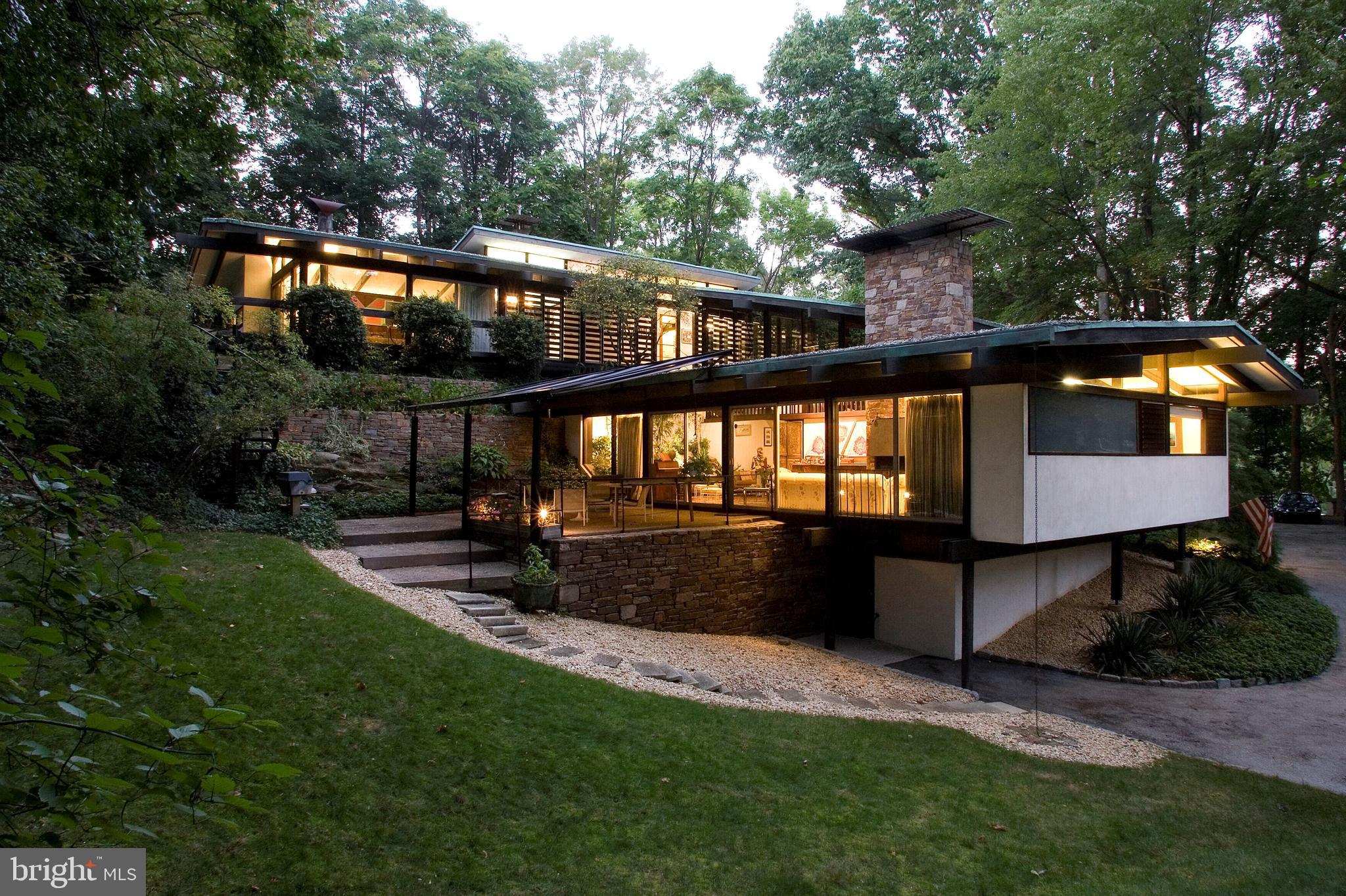 Nestled within an idyllic neighborhood, this mid-century marvel, created by Irwin Stein, stands as an iconic gem, boasting original installations that harken back to its timeless era. Offering between 5 bedrooms and 4 full baths alongside an additional half bath, this residence spans four meticulously finished levels. Nestled admist 1.2 acres of private surroundings with a large area for a playgound or outdoor enjoyment, the house seemlessly integrates with nature, is filled with natural light, and most rooms open to the outside.
Approaching the entrance, one is greeted by wide stone stairs leading to a bespoke entry door, flanked by sleek glass panels. Upon stepping inside, a tiled transverse foyer offers a tantalizing glimpse of the living room's captivating rock garden. Ascending the impressive catwalk staircase, which appears to float amidst expansive glass windows, one is enveloped in natural light pouring in from the southern and western exposures.
The fireside combination living and dining room exudes warmth and elegance, while sliding doors seamlessly connect to a covered terrace, perfect for alfresco dining amidst lush greenery and a charming ornamental pond. Adjacent to the dining area lies a kitchen straight out of a Mid Century Modern design book, retaining its original wood cabinetry and exceptional functionality.
Ascending the dramatic stairs leads to a sprawling primary suite, illuminated by full glass walls and complemented by a cozy corner woodburning fireplace. The sleeping area is delineated by custom-made Walnut furnishings, leading to an open closet and built-in desk, before culminating in the primary bath. Sliding doors reveal a deck overlooking the picturesque pond and gardens below, with a circular stairway, sourced from Philadelphia's famed Shubert Theater, providing access to the upper lawn and pool area.
On the same level, three additional bedrooms, two full baths, and a convenient laundry area cater to family and guests alike. Descending to the lower levels, one finds two distinct spaces, with the first serving as a potential fifth bedroom complete with a full bath and front exit. The second space, originally purposed as a recreational area, offers ample room for leisure activities or additional study and office space.
Located within the award-winning Lower Merion school district and just minutes from the city center, this residence transcends mere functionality, standing as a true work of art. A testament to architectural excellence, it epitomizes the mid-century aesthetic while offering unparalleled comfort and style. Viewing is available by appointment only.