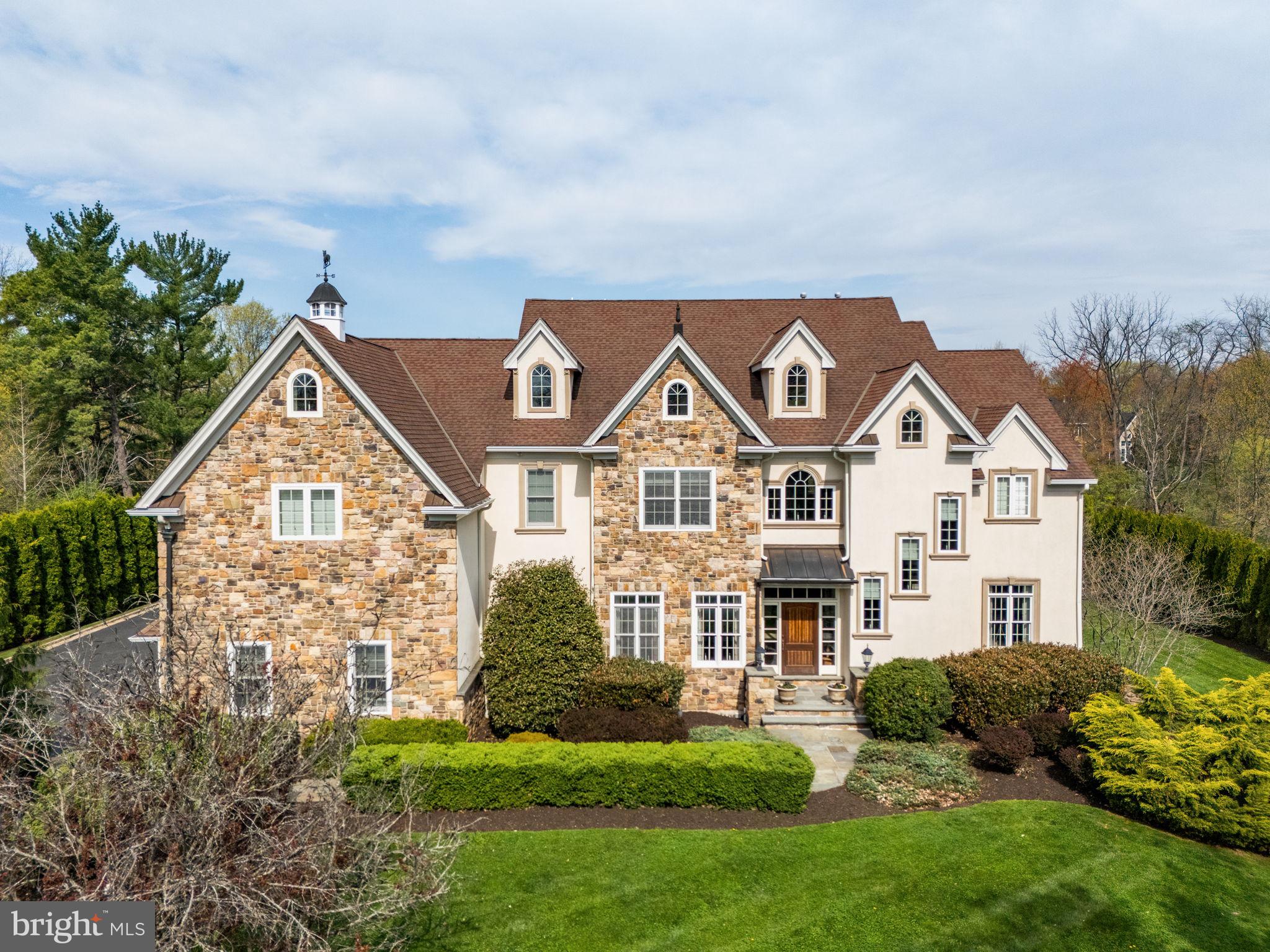 Set behind a private gated entrance, this remarkable estate is being offered for the first time and is gracefully situated on over an acre of meticulously manicured, tree-lined grounds. A cobblestone-lined driveway, enhanced by reclaimed European cast iron streetlights lead to the impressive residence, which offers more than 9,200 square feet of finished living space across four expansive levels. Upon entry, you are welcomed by a soaring foyer adorned with intricate trim work, crown molding, substantial baseboards, and elegantly detailed archways with corbel accents. The foyer is flanked by a formal dining room on one side and a refined living room on the other, while a private study adds both function and sophistication. Designed for grand entertaining as well as everyday living, the main level is anchored by a dramatic two-story great room featuring an impressive natural stone fireplace. The chef’s kitchen is equally exceptional, showcasing custom cabinetry, two oversized islands, a six-burner AGA range, a panel-ready Sub-Zero refrigerator, and a bright breakfast area that opens to an oversized Trex deck. This level is further enhanced by a butler’s pantry, a secondary powder room, and a mudroom with access to the attached three-car garage. Ascend the grand staircase to a striking catwalk that overlooks both the great room and foyer below. The second level offers four generously proportioned bedrooms, each with its own ensuite bath. The primary suite is a true sanctuary, appointed with a tray ceiling, exquisite millwork, private balcony access, two walk-in closets with custom built-ins, and a spa-inspired bath featuring cathedral ceilings, dual vanities, heated floors, a soaking tub, and a unique wraparound shower. The third floor adds exceptional versatility, offering a full bath and three additional bedrooms, ideal for extended family, guests, or flexible lifestyle needs. The lower level is thoughtfully finished and presents an extraordinary collection of amenities, including a media room with an additional stone fireplace, a speakeasy-inspired bar with a custom wood bar top, an additional bedroom, a theater room, a fitness area sporting a steam shower and a second private workspace with a dedicated entrance. Equally impressive, the backyard evokes a private resort setting, complete with a stunning in-ground saltwater pool, an outdoor fireplace, and a beautifully appointed outdoor kitchen with stainless steel appliances and granite countertops. Additional highlights include custom windows, a central vacuum system, dedicated HVAC zoning for each floor, a chandelier lift, a built-in sound system, and a host of other custom features throughout. Ideally located just moments from Ambler Borough, the property enjoys convenient access to some of Montgomery County’s most notable dining and shopping destinations, major transit routes, and the award-winning Wissahickon School District.