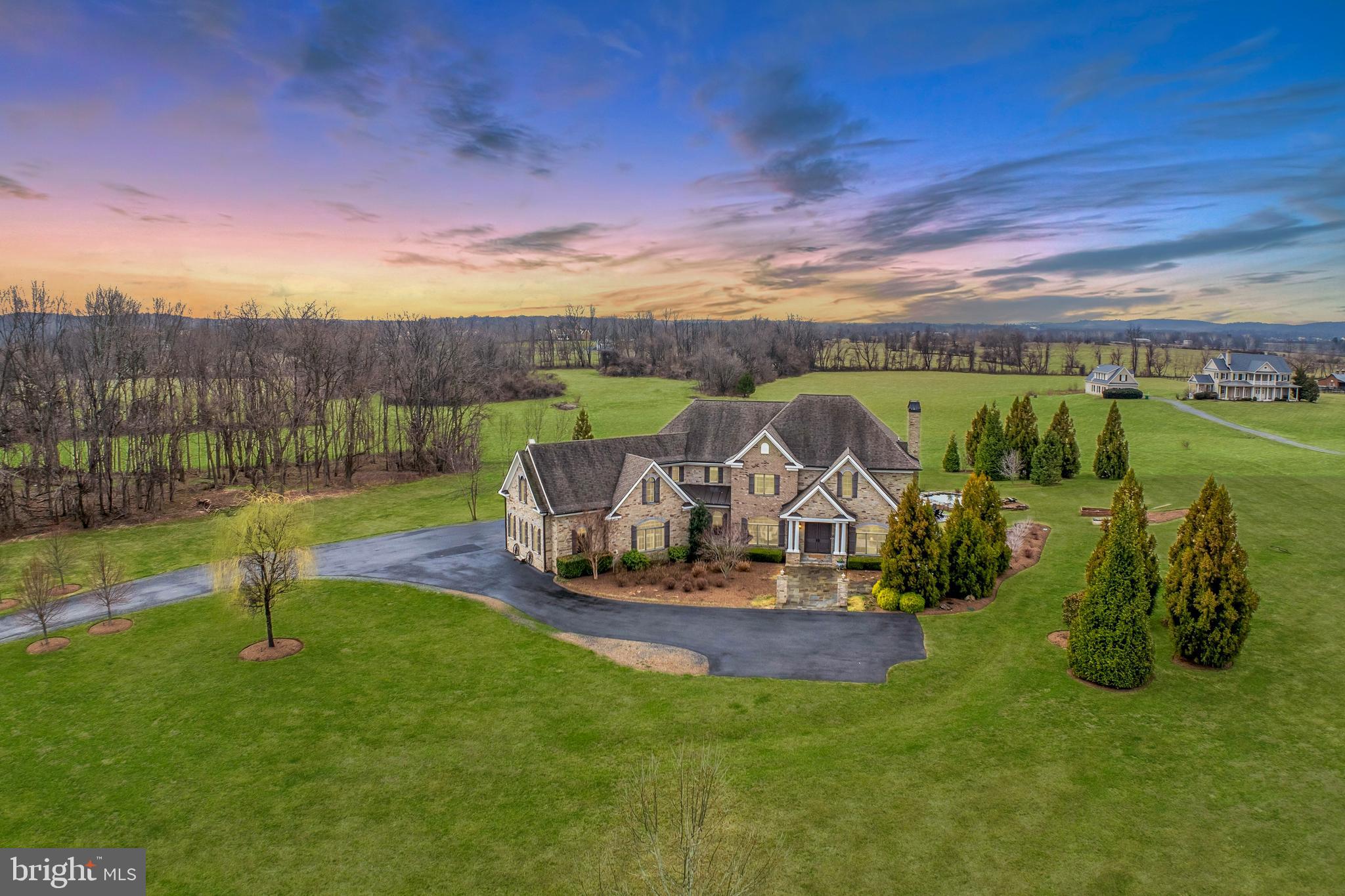 This romantic French Country revival, set within coveted Lee’s Crossing, unfolds gracefully from its bucolic surroundings, where provincial architecture is framed by flowering trees and seasonal blooms. Built in 2017, the home captivates with the chic sensibility of nearby Lucketts shops, showcasing over 5,500 square feet of open living finished with wide-plank hardwood floors, tailored built-ins, and elevated ceiling heights. Delicate chandeliers gently punctuate thoughtfully crafted spaces for gathering, dining, and retreating, while expansive windows frame pastoral vistas and sweeping sunset views like living works of art. In the airy chef’s kitchen, professional-grade appliances pair beautifully with cloud-hued cabinetry, stone countertops, and a nostalgic farmhouse sink. From the island, add the finishing touches to meals as guests select a bottle of local vino from the wine refrigerator. Outdoors, the covered grill space invites relaxed game nights and late-evening conversations around the firepit, with s’mores-ready serveware close at hand. Overlooking 13.8 sweeping acres, the landscape inspires visions of a future barn, a summer-ready pool, or open lawn games with a four-legged companion. Perhaps the home’s most striking feature, the central staircase serves as both anchor and destination, leading to restful reading nooks and casually sophisticated spaces designed to tuck away life’s essentials. Five sun-filled bedrooms each connect to high-end baths featuring rain showers, refined hardware, and serene spa-like palettes. A main-level suite comfortably accommodates long-term guests, while upstairs, the owner’s retreat offers a wet bar, dual custom dressing closets, and an indulgent soaking space designed for unwinding. Effortlessly impressive, the upper level also reveals a creative, popcorn-perfect bonus room, alongside a three-car garage and a thoughtfully designed mudroom finished with herringbone tile—setting the tone for cheerful departures and everyday adventures. Just moments from neighborhood horse trails, this exceptional home is surrounded by rural beauty while remaining conveniently close to bustling Leesburg, charming Lucketts, local farmers markets, and all that greater Loudoun County has to offer.