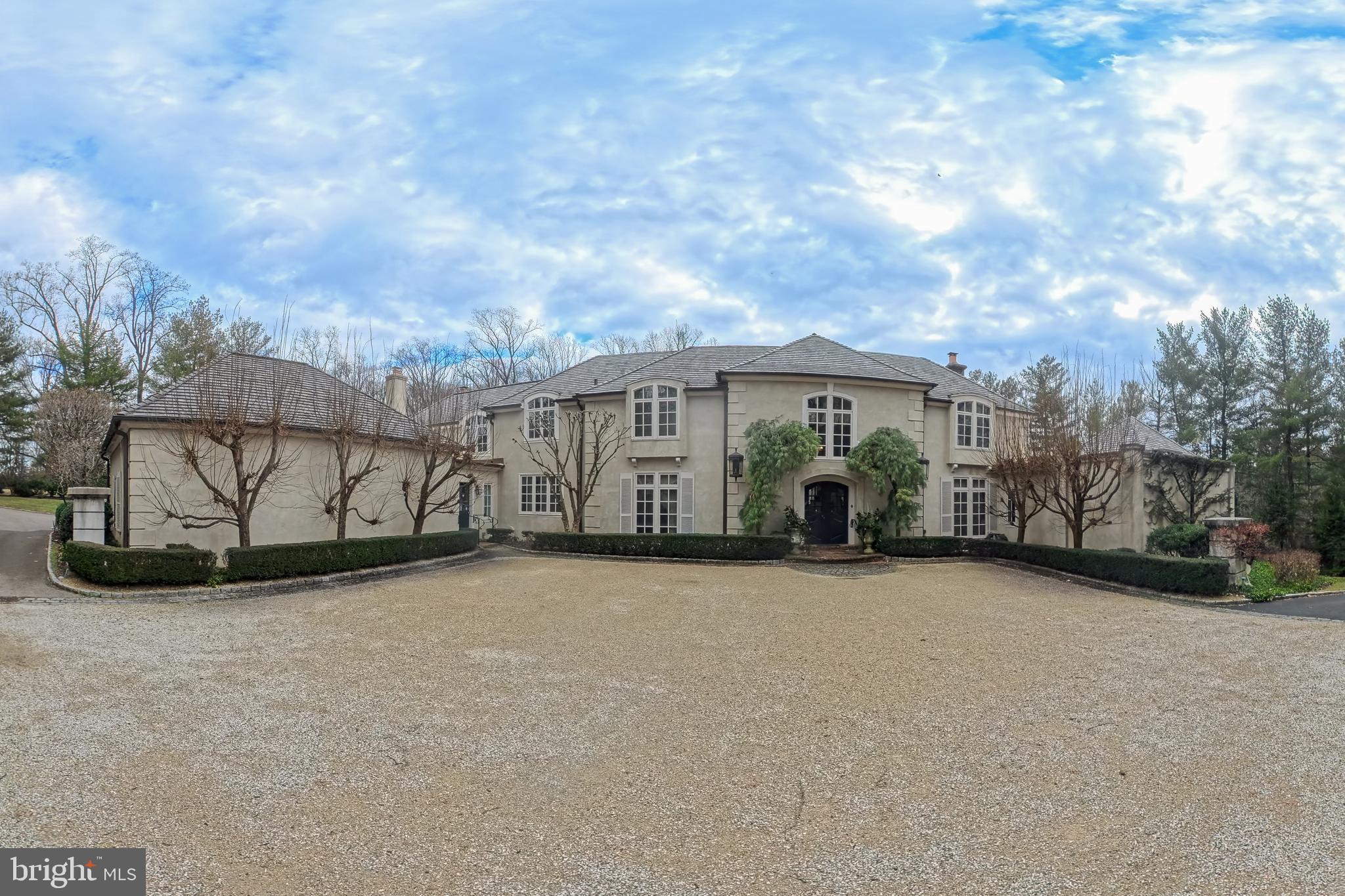 Situated in the heart of Gladwyne, this distinguished residence offers timeless architectural elegance and thoughtfully curated indoor and outdoor living spaces. A grand double-door entry opens to a marble-tiled foyer with soaring ceilings and a sweeping staircase.
The formal living room is highlighted by a dramatic two-story ceiling and expansive floor-to-ceiling windows. A beautifully appointed office features custom wood built-in bookshelves and a fireplace with a striking black marble surround. A second living room, ideally located near the kitchen, is rich in character with cathedral ceilings accented by wood beams, custom wood paneling, detailed crown molding, a fireplace, and a built-in bar alcove, creating an inviting setting for entertaining.
The kitchen is equipped with custom cabinetry, granite countertops, a Dacor gas cooktop, and a         double-door custom Sub-Zero refrigerator. The dining room is a true showpiece, featuring a hand-painted mural ceiling, dual chandeliers, and oversized doors that open to the back patio.
The first-floor primary suite offers a serene retreat with direct backyard access and two ensuite bathrooms, including an updated spa-like bath with a walk-in shower, bench seating, and overhead shower, as well as a second bath with a soaking tub and walk-in shower. Upstairs, six additional bedrooms are complemented by multiple full bathrooms, including several updated baths.
The finished basement adds flexible living space with a recreation area, home gym, and full bath. Outdoor living is elevated by a pavilion-covered patio designed for year-round enjoyment, featuring a substantial brick fireplace with built-in TV, a pool, and professionally landscaped grounds. A three-car garage completes this exceptional Gladwyne offering.