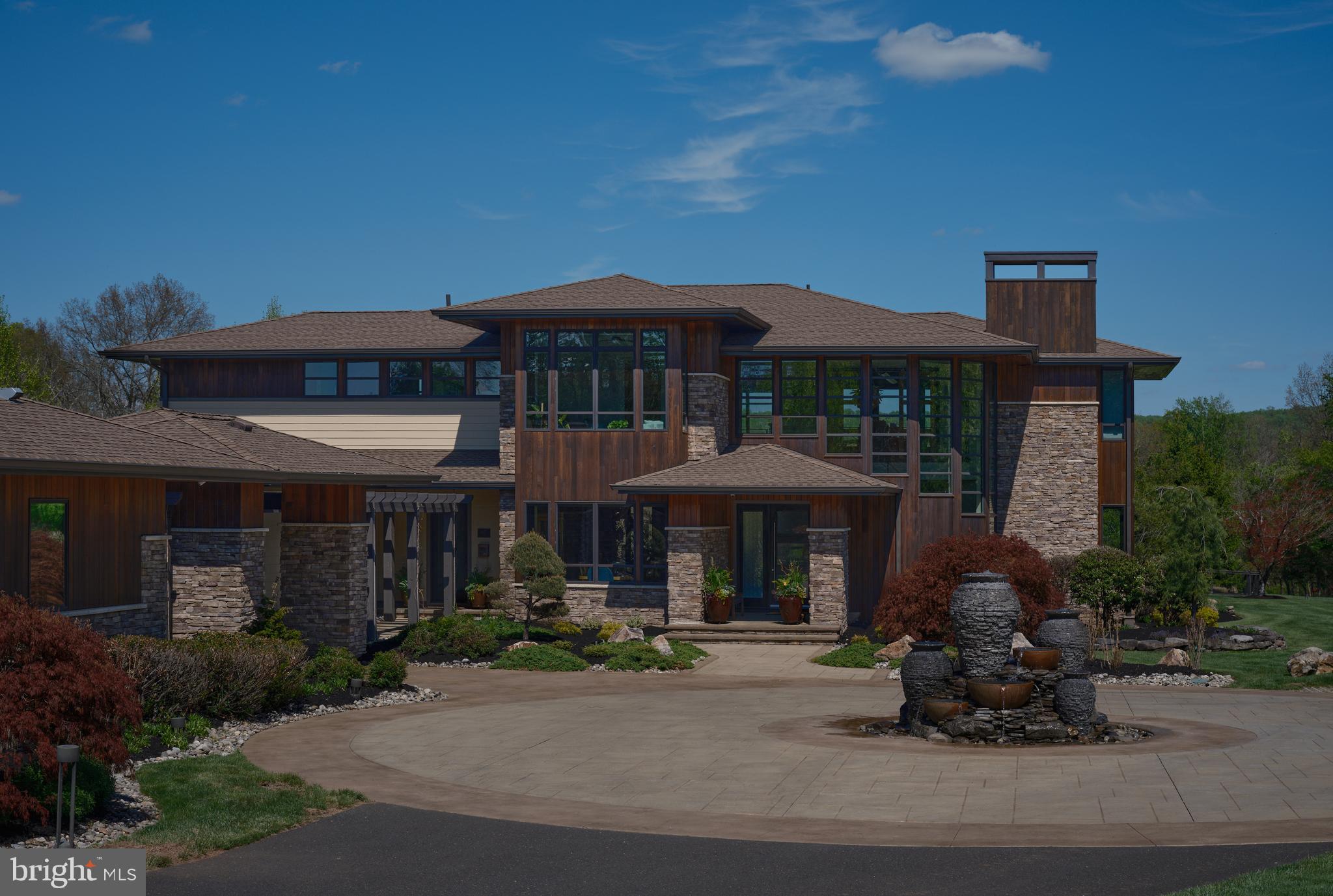 Down a private, tree-lined drive, this extraordinary 6-bedroom, 6 .5-bath estate—complete with an attached one bedroom guest house—is gracefully set on 10.8 serene acres. A breathtaking approach unfolds to a circular driveway, highlighted by a stunning custom water feature.  Inspired by the iconic designs of Frank Lloyd Wright, this architectural masterpiece is thoughtfully integrated into its natural surroundings, offering an exceptional balance of sophisticated entertaining and peaceful everyday living. Hallmarks of Wright’s influence are evident throughout, including expansive walls of glass, custom interior stonework, cantilevered rooflines, and dramatic windows that seamlessly bring the outdoors in. Natural materials such as cork flooring and radiant-heated ceramic tile further enhance the home’s organic aesthetic.  A striking two-story foyer welcomes you with a sweeping spiral staircase, custom fabricated iron railing, and a dramatic floor-to-ceiling stone wall that draws the eye into the impressive two-story great room beyond, where uninterrupted views of the rear grounds await. To the right of the double-door entry, a private wing offers a sitting room, bedroom with full ensuite bath, and an additional powder room—ideal for guests or multigenerational living.  The opposite wing reveals a spectacular open-concept living and dining space with soaring 21-foot vaulted ceilings, floor-to-ceiling windows, and a granite-faced gas fireplace. The chef’s kitchen is beautifully appointed with dual islands, custom cherry cabinetry, quartzite countertops, and premium appliances including Thermador, Miele, and Fisher & Paykel, along with a built-in coffee system and dumbwaiter.  The adjoining breakfast room is bathed in natural light, with two walls of soaring windows and direct access to a covered stone patio—perfect for morning coffee or al fresco dining. Designed with entertaining in mind, three separate walk-in pantry rooms off of the kitchen with custom cabinetry provide exceptional storage for appliances, serving pieces, and household essentials. This wing flows seamlessly into a spacious mudroom and laundry suite, complete with a ceramic dog-grooming tub and access to the attached two-car garage.  Upstairs, the primary suite serves as a private sanctuary, featuring a gas fireplace, a cozy sitting area overlooking the pool and great room, and a luxurious ensuite bath with dual vanities, a custom two-person steam shower, jetted whirlpool tub, radiant heated floors, and a Toto toilet. Expansive dual walk-in closets complete the suite. Two additional bedrooms, each with ensuite baths, and a well-appointed office with custom cherry double wraparound desks and extensive built-ins provide both comfort and functionality.  The walk-out lower level is designed for recreation and relaxation, offering a full kitchenette with custom cherry cabinetry and granite countertops, a bar, multiple seating areas, home gym, and two guest bedrooms with ensuite baths. Oversized doors open to one of three private patios, extending the living space outdoors.  The rear grounds have been transformed into a private resort, ideal for both grand-scale entertaining and everyday enjoyment. A custom kidney-shaped saltwater pool with waterfall, tanning ledge, and spillover spa is surrounded by an expansive stamped concrete patio and lush professional landscaping. A thoughtfully designed pavilion features a full outdoor kitchen, a bar, TV lounge with stone fireplace, rooftop observation deck, full bath with changing area, and a dedicated laundry room. The guest house connects via a breezeway and offers additional living space with a kitchen, bedroom, office, and private patio, plus an attached two-car garage.  Additional highlights include a solar field with sun-tracking capability and a generator, ensuring efficiency and peace of mind.  Ideally located on a bucolic road in Bucks County and within the awarding winning Council Rock School District.