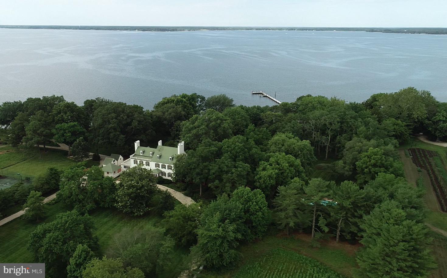 Wilderness Estate- This quintessential Eastern Shore farm is located along the Choptank River in Trappe, Maryland. Comprised of 47± acres, with original structures that are impeccably well-kept and improved with modern amenities, The Wilderness provides a unique opportunity to enjoy the best of the Eastern Shore.  Surrounded by open space and preserved farmland with proximity to Oxford, Easton, Cambridge, and St. Michaels, The Wilderness is sure to please the discerning buyer.  
Sited among large waterfront estates, The Wilderness enjoys privacy and a commanding presence with old-growth trees, broad southerly views, and deep water.  The scope of improvements includes the primary residence, waterside guest cottage, beach house, pool, pier, tennis court, early 20th-century barn, and a host of accessory structures for life on the farm.  Visit the virtual tour for an inside look.

The main house has great historical significance not only for the original owner’s grandson who lived there and served two terms as the Governor of Maryland but also for its place in the development of Maryland’s domestic architecture, as noted on two markers from the Maryland Bicentennial Commission and the Maryland Historical Society. 
The smaller and original of the two brick wings dates from the late 18th century and the larger wing from the early 19th century. The house’s architecture is unusual in that the 19th-century wings telescope up, instead of down, from the original wing. The white brick, black shutters, and historical olive green trim color are a serene palette against the house’s majestic specimen trees around the property, including a gigantic Sycamore that was at one time, the largest of its species in Maryland. 
The long white gravel drive under the sheltering canopy of mature trees passes by the barn and other outbuildings then curves around the front of the house. Brick steps lead up to the front porch of the early 19th century wing opening into a gracious foyer and stair hall with an elegant elliptical arch that rests on pilasters at the side walls to separate the public from private areas of the house. A French door at the rear of the foyer frames a vista of the lawn and Choptank River. To the side of the foyer are two identical parlors at the front and rear corners of the house. Both rooms have magnificent fireplace mantles and surround; one is a 1740 hand-carved mantel by noted English artisan woodcarver Grinnling Gibbons and the other is a 1780 mantel surrounded by the famous English architect and artisan, Robert Adam. 
The main floor of the 18th-century wing contains a spacious living room, dining room, original kitchen, and larger kitchen, pantry, two powder rooms, and a stunning sunroom connecting to a terrace along the rear of the house overlooking the Choptank River. Part of this wing was the final addition to the original home.
The second floor of the 19th-century wing contains two bedrooms over the two parlors below with fireplaces and walk-in closets and two baths. The 18th-century wing and later addition contain an artist’s studio with cabinetry and sink that has a myriad uses– TV room, arts and crafts room. The primary suite is located at the rear corner of this wing. It has a fireplace flanked by single windows and another window on the rear wall for panoramic views of the mature landscape and the Choptank River. Two baths complete the suite, with one bath off a hall for access from the art studio. 
Two stairs access the attic level; one in the 18th-century wing leads to two bedrooms and one bath. The main stair in the 19th-century wing leads to a large game room and another cozy bedroom ensuite.