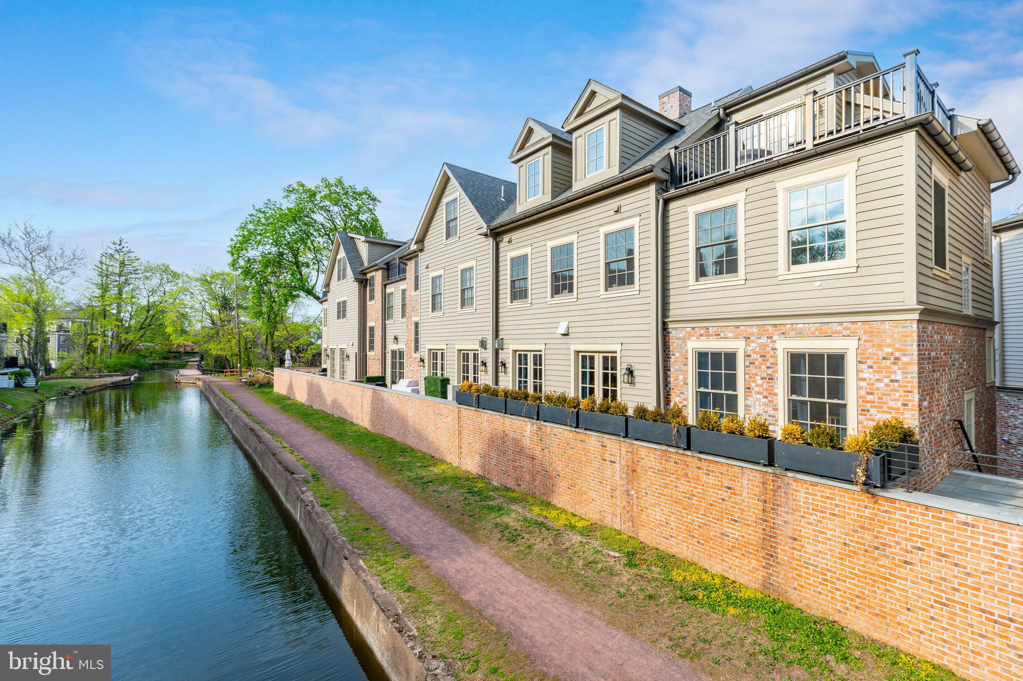 Welcome to The Falls at New Hope, an exclusive collection of luxury townhomes designed by award-winning architect Ralph C. Fey, where sophisticated design meets the natural beauty of Bucks County. Perfectly positioned along the historic Delaware Canal and just moments from the heart of downtown New Hope, this exceptional canal-front residence offers a rare opportunity to experience refined, four-level living in one of the area’s most coveted communities.

Thoughtfully designed with both function and elegance in mind, this three-bedroom, three full and two half bath home unfolds across four beautifully appointed levels, all connected by a private elevator for seamless accessibility. From the moment you enter the welcoming foyer, the home sets the tone with clean architectural lines, wide-plank hardwood flooring, and a soft, curated palette that enhances the natural light throughout.

The main living level is designed for elevated everyday living and entertaining, featuring a stunning open-concept layout that seamlessly connects the kitchen, dining, and living spaces. The chef’s kitchen is a true showpiece, offering custom cabinetry, sleek stone countertops, a large center island with seating, and a full suite of high-end appliances including double wall ovens, dual wine refrigerators, and an impressive 60-inch side-by-side refrigerator and freezer. A dedicated water filtration system with separate spout adds both convenience and refinement. The space flows effortlessly into the living area, where a striking linear fireplace with modern surround creates a warm yet sophisticated focal point, complemented by expansive windows and access to outdoor space overlooking the canal. Thoughtful enhancements such as central vacuum, including discreet in-wall and under-cabinet access points, further elevate everyday functionality.

The third level offers two generously sized bedroom suites, each with its own beautifully finished en-suite bath. One of the bedrooms is complemented by a dedicated office enclosed by dramatic 8-foot glass and steel doors, creating a striking and functional work-from-home environment. The adjoining bath is a standout, featuring rich green tile, a glass-enclosed shower, and designer fixtures that add depth and character to the space. Custom-built closets throughout this level provide both style and organization.
The entire top level is devoted to the primary suite, offering a true private retreat. This expansive space features vaulted ceilings, abundant natural light, and access to a private terrace overlooking the canal. A wet bar enhances the level, creating a perfect setting for morning coffee or evening unwinding. The spa-inspired primary bath is beautifully appointed with dual vanities, elegant finishes, and a sculptural soaking tub, along with a glass-enclosed shower for a complete, luxurious experience. The terrace is equally impressive, featuring planter boxes with an integrated auto-watering system, allowing for effortless outdoor living and seasonal enjoyment.

Additional highlights include an attached two-car garage with built-in storage and finished flooring, a hot water recirculation system for enhanced pressure and efficiency across upper levels, a comprehensive ADT security system with multiple live camera feeds, and meticulous craftsmanship throughout that reflects the architectural integrity of the community.

Residents of The Falls at New Hope enjoy a low-maintenance lifestyle with direct access to the Delaware Canal path and close proximity to New Hope’s vibrant dining, shopping, and cultural scene, including the historic Bucks County Playhouse. Across the river, Lambertville adds even more charm with its galleries, boutiques, and scenic views. Unit #1 represents a rare offering of canal-front luxury, where thoughtful design, elevated finishes, and modern conveniences come together in one exceptional home.