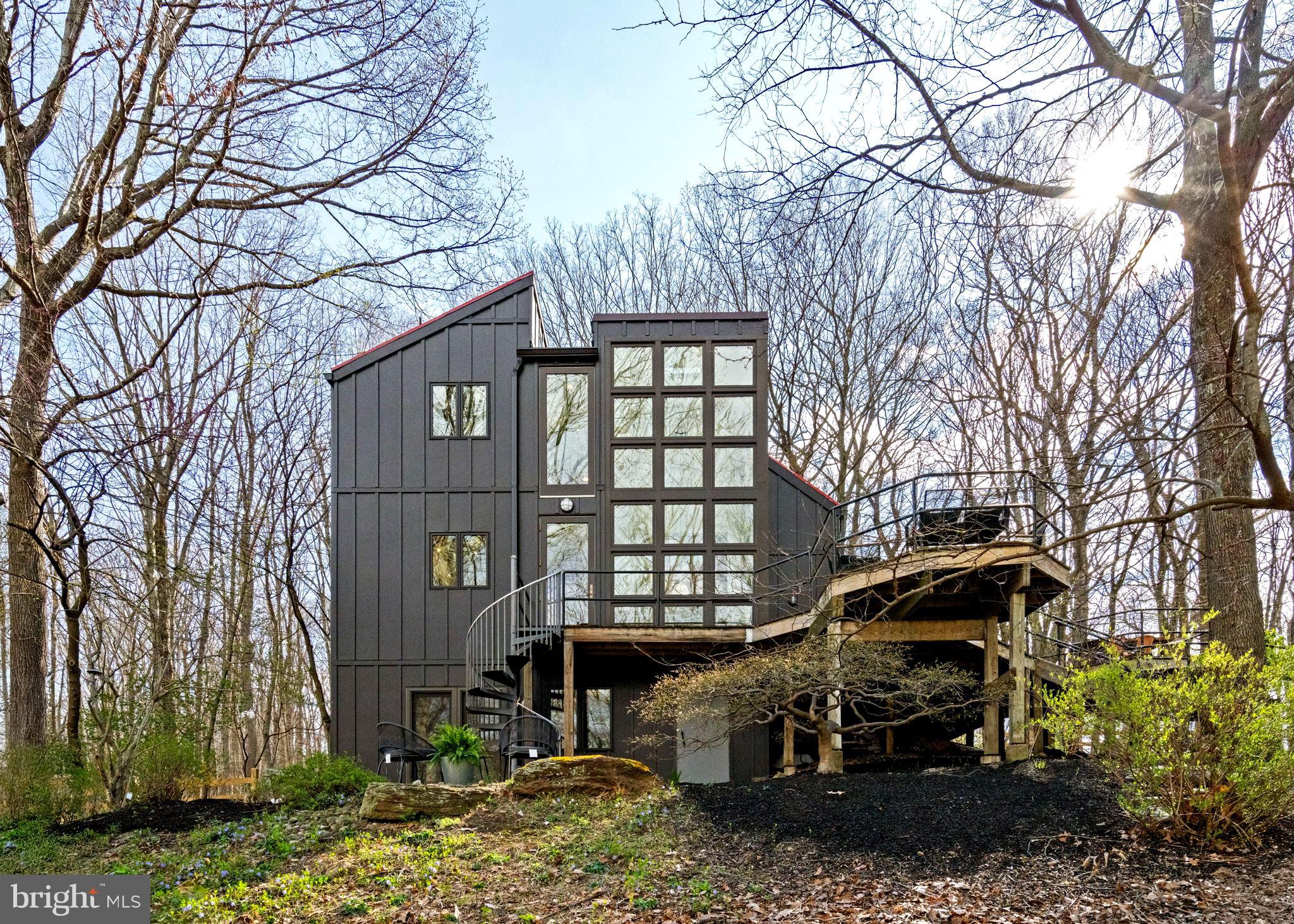 Breathtaking architecture on three wooded acres provides a residence as equally well-suited for entertaining as it is for tranquil everyday living in this Bucks County treasure. The Scandinavian influence is evident in the full walls of glass and dramatic windows that bring the outside in, a central chimney that provides a fireplace on every floor, and the use of natural materials including slate and oak floors. Lovely custom details with a touch of whimsy grace the interior, and the current owners have made extensive improvements. The main floor offers a spacious foyer entrance, library with skylight, powder room and sitting room all on one side of a double fireplace with newly installed woodstove insert. On the other side is the showstopping open living and dining space with vaulted ceilings, floor-to-ceiling windows, custom conversation pit with integrated storage that seats a crowd, and a just-installed wall of custom 12-foot glass bifold doors that allow the separation between the inside and the expansive Ipe wood deck with multiple entertaining areas to all but disappear. The corner breakfast room has two walls of floor-to-double-story-ceiling windows, from which the current owners eliminated a wall and added a breakfast bar to make it more open to the kitchen, where the Scandinavian design principles really come to life. Functional details like a prep sink, pot filler and wine fridge are integrated into the understated warmth and luxury of oak flat-front cabinets alongside fresh quartz countertops and glazed ceramic tile backsplash. The second story has a large central sitting room with fireplace overlooking the living room with a newly renovated terrazzo tile bathroom, two bedrooms, and an office with French doors to a balcony. The third floor is the peaceful primary suite, with a fireplace and sliders to a balcony that spans the entire side of the house and overlooks the wooded hillside and pool area. Soak among the treetops in the gorgeously remodeled en suite, where the tub sits beneath a span of oversized sloped windows, or enjoy the same view from the curbless shower with fluted marble surround. The delightful walkout lower level has a wet bar, a large den with oversized French doors to a cobblestone patio, a guest suite with a newly renovated bathroom, and access to the laundry room. Just as stunning as the home itself is the newly plastered pool and spa with flagstone pavers, which are framed by a decorative wall on one side and an expanse of woods on the other. Other exterior upgrades include new elegant black Hardie board and batten siding and a fence encompassing a large area of the yard and pool. In a bucolic Bucks County setting and down a private road, the home is away-from-it-all yet a short drive to Doylestown, New Hope, and Lambertville, making it both a desirable year-round residence and an attractive getaway for Philadelphians and New Yorkers alike.