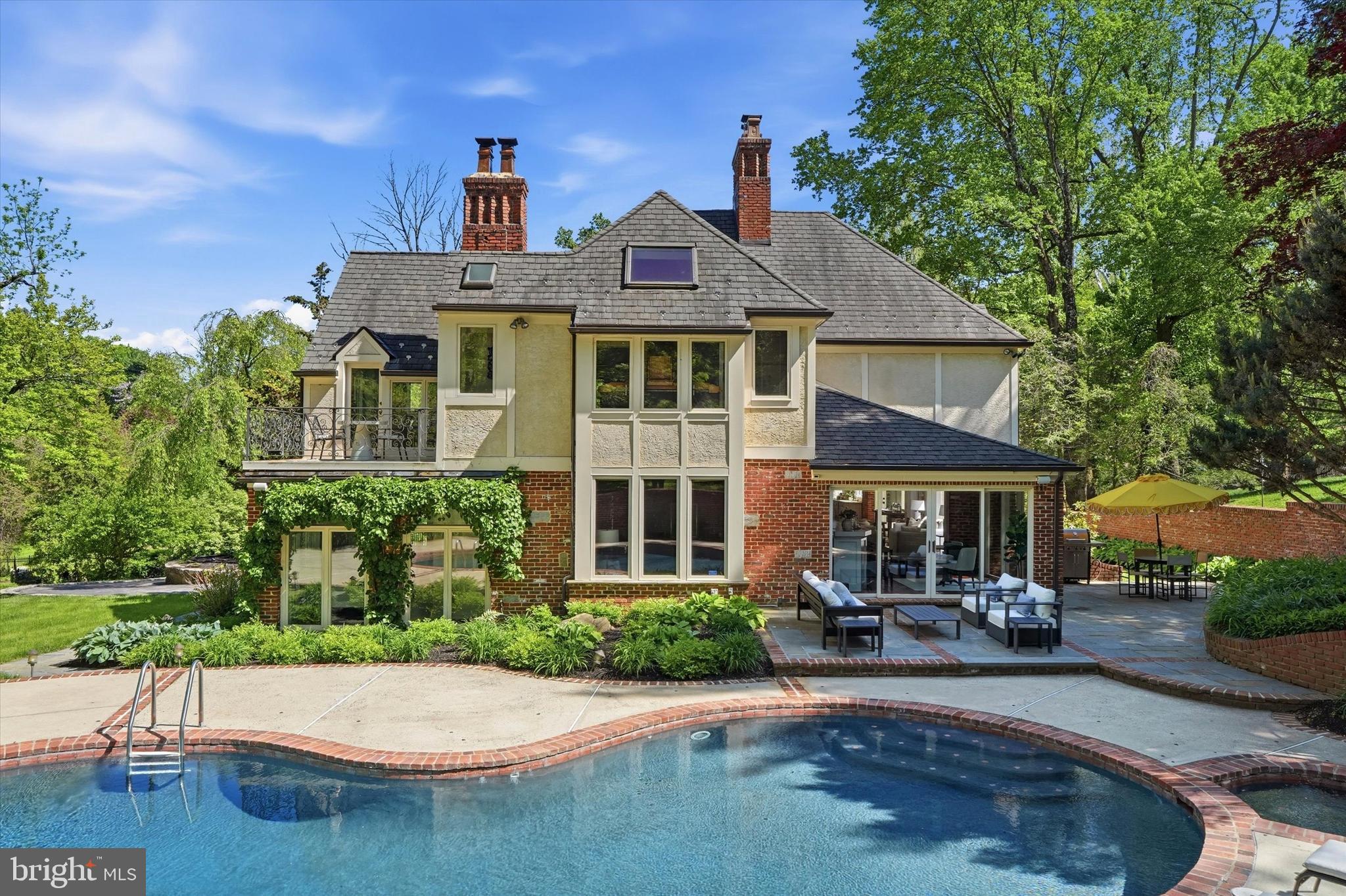 Set beyond an iron gate on 1.9 picturesque acres along a private road, 1100 Green Valley is a distinguished Bryn Mawr estate offering the privacy, presence, and enduring character of a classic Main Line residence. Approached by a circular drive & framed by sweeping lawn, mature landscaping, & beautifully designed outdoor spaces, this handsome stone and brick Tudor-style home makes an unforgettable first impression. The grounds are lush and layered, with dogwood, holly, Japanese magnolia, pine, and mature evergreens providing year-round color.
Inside, the residence has been thoughtfully refreshed while preserving the architectural detail, warmth, and craftsmanship that give it such distinction. A new roof, updated lighting throughout, freshly painted, and enhanced baths create a polished, cohesive feel, while the home’s timeless character remains beautifully intact. The floor plan offers exceptional natural flow, with formal and informal spaces that connect effortlessly for elegant entertaining, everyday living, and quiet retreat.
A gracious foyer introduces the home’s timeless appeal and opens to the step-down living room, where hardwood floors, crown molding, a fireplace, and an adjoining sunroom, creating an elegant yet inviting gathering space. Nearby, a richly paneled office with closets, built-ins, and its own fireplace offers a refined work-from-home setting and could also serve as a 5th bedroom. A full hall bath and generous coat closet add convenience to this wing of the main level.
Across the foyer, the formal dining room provides a beautiful setting for hosting, enhanced by built-in cabinetry and a sophisticated sense of style. The kitchen blends texture, charm, and function with vaulted beamed ceilings, exposed brick, and heated floors for added comfort, along with raised-panel cabinetry, marble countertops, a center island with seating, instant boiling water, and high-end appliances including paneled SubZero refrigerators and a Viking range with hood. A brick archway leads to the sun-filled breakfast area, where skylights and direct access to the grounds create a seamless connection to the outdoors.
The FR is warm and inviting, featuring herringbone hardwood floors, a brick FP with built-ins, and an adjoining sunroom with a built-in wet bar. These spaces open beautifully to the patios and rear grounds, making the home exceptionally well-suited for indoor-outdoor entertaining. The main level also includes a spacious combined pantry and laundry room with custom cabinetry, wash tub, and excellent storage, along with a beautifully appointed powder room nearby. 3 HVAC systems, including 1st floor air filtration, further enhance comfort and livability.
Upstairs, the private primary wing feels like its own retreat, offering hardwood floors, a cathedral ceiling, private terrace, custom closet, and a spacious bath with steam shower, separate soaking tub, and heated floors. An open office or lounge area leads to two additional bedrooms, each with walk-in closets and ensuite baths.
A fourth bedroom suite, positioned just a half level above the two-car garage, includes its own full bath and offers wonderful flexibility for guests, an au pair, in-laws, or extended stays. The two-car garage is also wired for an EV charger, adding modern convenience to this classic estate setting. The partially finished lower level adds additional space, including a full bath, storage, and room for recreation, fitness, or hobbies.
The outdoor setting is truly extraordinary. Expansive patios invite outdoor dining, grilling, lounging, and entertaining, while the in-ground pool with hot tub offers a resort-like experience at home. A charming duck pond and waterfall bring a sense of tranquility and escape, and an outdoor fountain hookup provides the opportunity to further enhance the serene landscape. The gated entry is thoughtfully wired with its own cellular network antenna for added reliability and convenience.