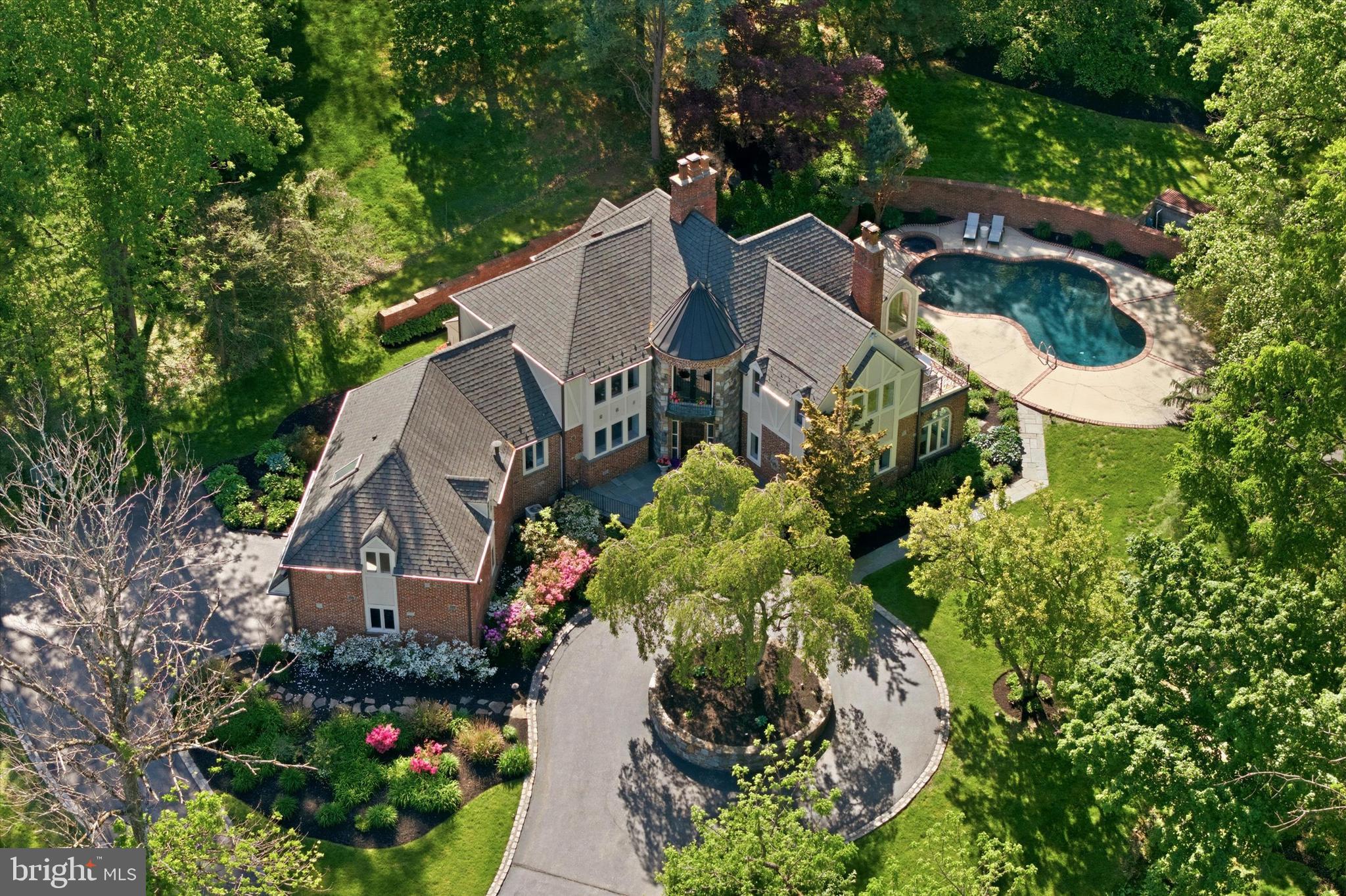 Set beyond an iron gate on 1.9 picturesque acres along a private road, 1100 Green Valley is a distinguished Bryn Mawr estate offering the privacy, presence, and enduring character of a classic Main Line residence. Approached by a circular drive & framed by sweeping lawn, mature landscaping, & beautifully designed outdoor spaces, this handsome stone and brick Tudor-style home makes an unforgettable first impression. The grounds are lush and layered, with dogwood, holly, Japanese magnolia, pine, and mature evergreens providing year-round color.
Inside, the residence has been thoughtfully refreshed while preserving the architectural detail, warmth, and craftsmanship that give it such distinction. A new roof, updated lighting throughout, freshly painted, and enhanced baths create a polished, cohesive feel, while the home’s timeless character remains beautifully intact. The floor plan offers exceptional natural flow, with formal and informal spaces that connect effortlessly for elegant entertaining, everyday living, and quiet retreat.
A gracious foyer introduces the home’s timeless appeal and opens to the step-down living room, where hardwood floors, crown molding, a fireplace, and an adjoining sunroom, creating an elegant yet inviting gathering space. Nearby, a richly paneled office with closets, built-ins, and its own fireplace offers a refined work-from-home setting and could also serve as a 5th bedroom. A full hall bath and generous coat closet add convenience to this wing of the main level.
Across the foyer, the formal dining room provides a beautiful setting for hosting, enhanced by built-in cabinetry and a sophisticated sense of style. The kitchen blends texture, charm, and function with vaulted beamed ceilings, exposed brick, and heated floors for added comfort, along with raised-panel cabinetry, marble countertops, a center island with seating, instant boiling water, and high-end appliances including paneled SubZero refrigerators and a Viking range with hood. A brick archway leads to the sun-filled breakfast area, where skylights and direct access to the grounds create a seamless connection to the outdoors.
The FR is warm and inviting, featuring herringbone hardwood floors, a brick FP with built-ins, and an adjoining sunroom with a built-in wet bar. These spaces open beautifully to the patios and rear grounds, making the home exceptionally well-suited for indoor-outdoor entertaining. The main level also includes a spacious combined pantry and laundry room with custom cabinetry, wash tub, and excellent storage, along with a beautifully appointed powder room nearby. 3 HVAC systems, including 1st floor air filtration, further enhance comfort and livability.
Upstairs, the private primary wing feels like its own retreat, offering hardwood floors, a cathedral ceiling, private terrace, custom closet, and a spacious bath with steam shower, separate soaking tub, and heated floors. An open office or lounge area leads to two additional bedrooms, each with walk-in closets and ensuite baths.
A fourth bedroom suite, positioned just a half level above the two-car garage, includes its own full bath and offers wonderful flexibility for guests, an au pair, in-laws, or extended stays. The two-car garage is also wired for an EV charger, adding modern convenience to this classic estate setting. The partially finished lower level adds additional space, including a full bath, storage, and room for recreation, fitness, or hobbies.
The outdoor setting is truly extraordinary. Expansive patios invite outdoor dining, grilling, lounging, and entertaining, while the in-ground pool with hot tub offers a resort-like experience at home. A charming duck pond and waterfall bring a sense of tranquility and escape, and an outdoor fountain hookup provides the opportunity to further enhance the serene landscape. The gated entry is thoughtfully wired with its own cellular network antenna for added reliability and convenience.