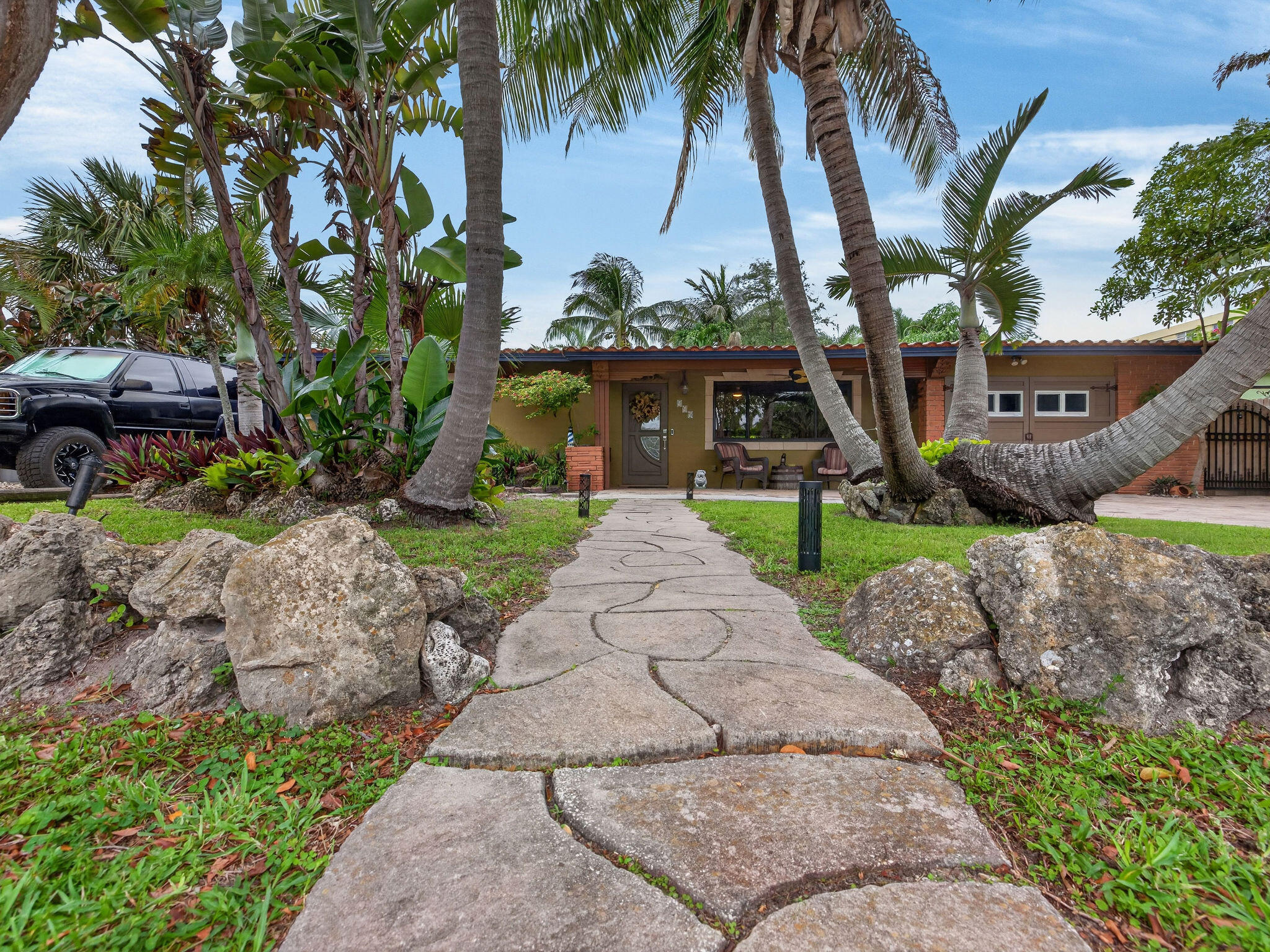 Tucked away behind a canopy of mature palms and lush tropical landscaping, this one-of-a-kind East Pompano Beach retreat offers the perfect blend of privacy, character, and resort-style living just minutes from the beach. The property welcomes you with a newly pavered driveway and custom stone walkway leading to the front entrance, while a gated side-entry covered parking area provides additional storage and flexibility for vehicles, watercraft, recreational equipment, or workshop space.  This lovingly maintained 3-bedroom, 2-bath residence features an open-concept layout with spacious living areas, a custom entertainment wall, tile flooring throughout the main living spaces, and seamless indoor-outdoor flow designed for both everyday living and entertaining. Throughout the home, unique  architectural details and custom design elements create a warm, inviting atmosphere, including arched openings, curved ceiling accents, custom woodwork, stone features, decorative niches, and thoughtfully crafted built-ins that add both charm and character.  Step outside into your own private oasis where a sparkling freeform pool is surrounded by expansive paver decking, tranquil koi ponds with cascading water features, winding stone pathways, and a curated tropical landscape that transforms the backyard into a true botanical retreat. Multiple outdoor gathering spaces, a covered pergola, fire pit area, and serene garden vignettes create an atmosphere rarely found in South Florida homes.  Adding exceptional versatility, the property includes a Master Suite which can also be used as an in-law suite complete with its own kitchen, bathroom, separate entrance, and dedicated mini-split A/C system.  The home also offers valuable peace of mind with a tile roof installed in 2007, a new water heater (2024), and two air conditioning systems replaced in 2021 and 2024. Additional benefits include irrigation supplied by the City of Pompano Beach's reclaimed water system, helping maintain the lush landscaping efficiently, as well as recently upgraded neighborhood sewer infrastructure with sewer line replacements already completed.  Situated on a generous 7,875-square-foot lot approximately one mile from the beach, this extraordinary property offers a unique combination of indoor comfort, outdoor beauty, and an unbeatable location near shopping, dining, golf, marinas, and all that coastal South Florida has to offer.