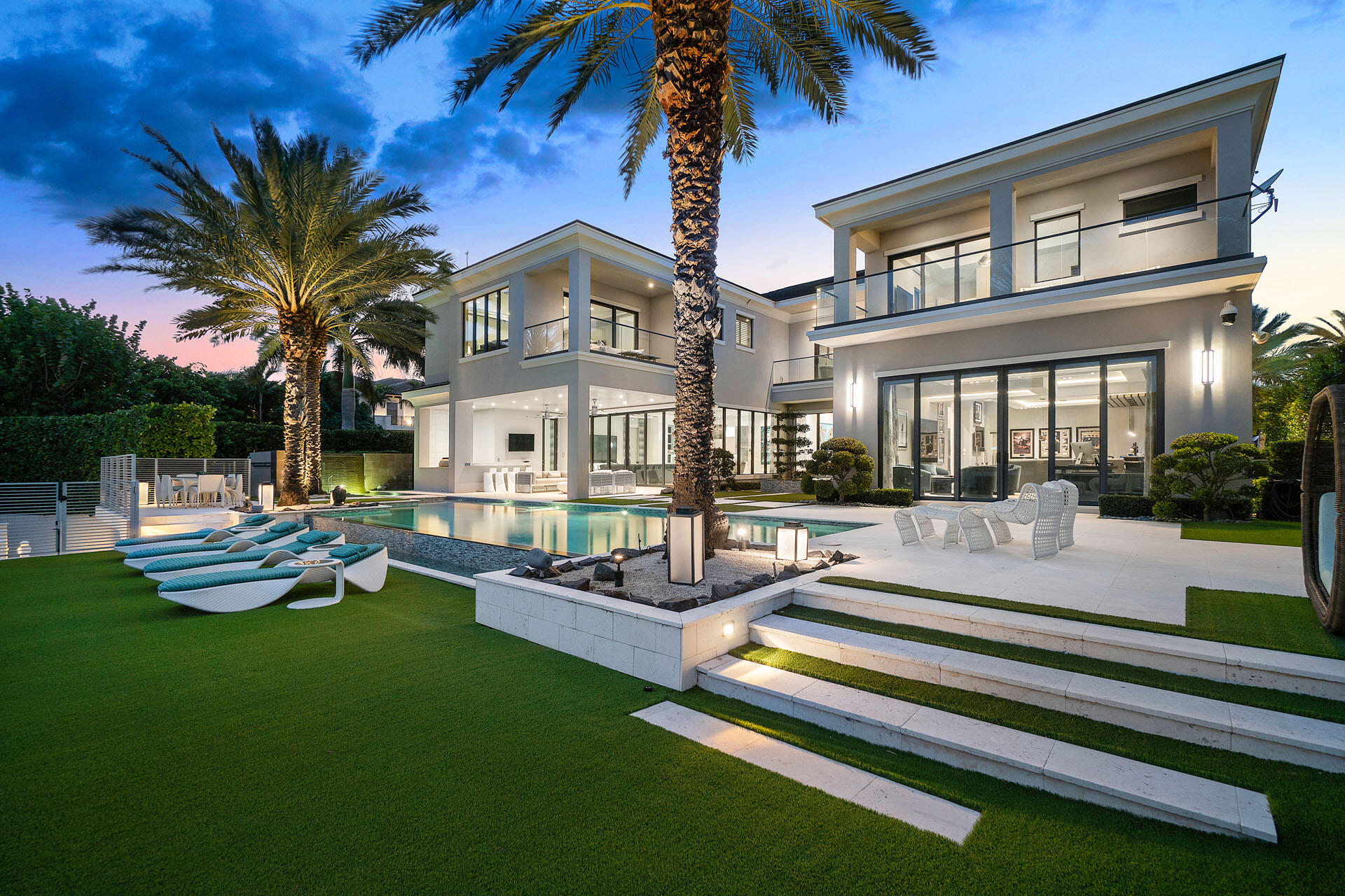 A commanding presence on Royal Palm Yacht & Country Club's Grand Canal, 290 South Maya Palm Drive is a masterpiece of modern coastal luxury by Bloomfield Construction. This waterfront estate spans over 9,000 square feet, offering five impeccably designed bedrooms and 6.2 baths, all crafted for those who demand the finest design and craftsmanship. A soaring two-story gallery foyer sets the tone for the home's exquisite interiors, where striking agate accents, floor-to-ceiling marble, and recessed architectural lighting create an ambiance of understated grandeur. The estate's expansive club room, featuring a luminous resin bar, is designed for lavish entertaining, while motorized sliding glass doors seamlessly connect indoor and outdoor living. The resort-inspired backyard is a private oasis, with a zero-edge infinity pool, separate spa, and elegantly appointed lanai, all overlooking 110 feet of prime water frontage. Glass rail balconies with turf floors provide a sleek vantage point to take in the breathtaking surroundings. The primary suites, each a private sanctuary, boast boutique-style dressing rooms and spa-like baths. A four-bay garage with car lift capability completes this estate, ensuring the home caters to lifestyle and legacy. Don't miss this unparalleled opportunity to own a statement residence in one of South Florida's most prestigious enclaves.