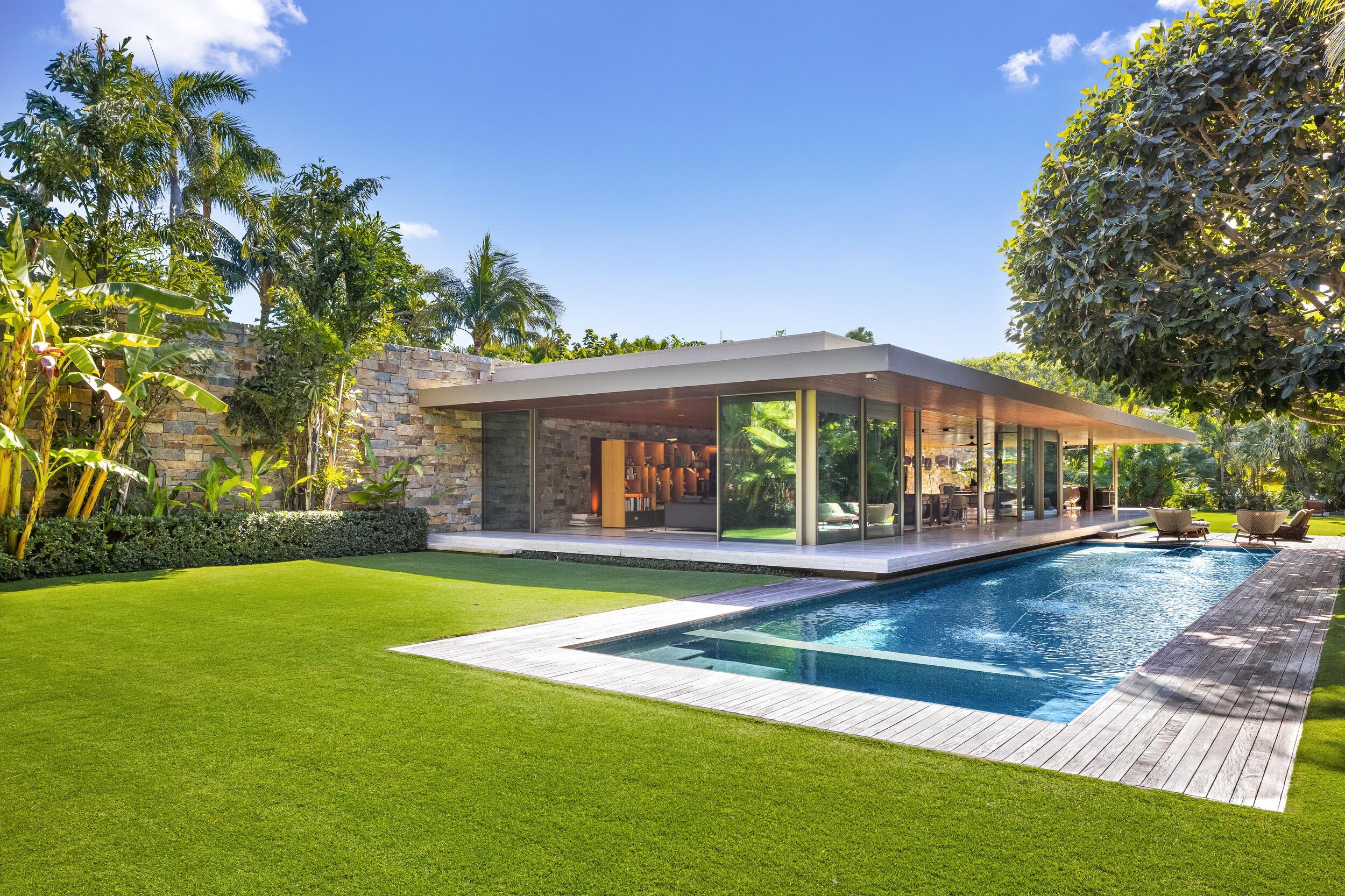 A stunning modern estate set on a rare, oversized 40,000± square foot lot, this exceptional one-story residence is a masterwork by award-winning modernist architect Marcio Kogan of Studio mk27, conceived to blur the boundaries between indoor and outdoor living. The home offers four bedrooms, five full baths, and two powder rooms, arranged within a thoughtfully curated floor plan that balances openness with privacy. Expansive floor-to-ceiling Vitrocsa glass walls flood the interiors with natural light and allow the living room to transform effortlessly into an open-air pavilion, overlooking lush, manicured gardens. Architectural details are both refined and intentional: teak rods line the ceilings, while travertine flooring extends seamlessly from interior to exterior, visually and physically uniting the spaces. The indoor living room, fully enclosed in glass, opens to tranquil garden views, while the outdoor living room is anchored by a dramatic metal fireplace suspended from the ceiling, creating a striking focal point.
A sculptural Virginia flagstone wall artfully separates the social areas from the private wing, which includes the bedrooms, kitchen, office, and media room. The primary suite is a serene retreat, featuring an oversized walk-in closet, dual baths, and peaceful garden vistas.
The exterior spaces are equally compelling. Loggias, a resort-style pool area, and lush tropical landscaping create a private outdoor oasis, enhanced by a dramatic living wall that serves as both an artistic statement and a unifying design element between inside and out.
The residence is further elevated by impeccable interior design by Diana Radomysler and landscape architecture by Keith Williams of Nievera Williams Design. Recognized for its architectural significance, the home is the Winner of the Preservation Society's 2020 Schuler Award.
This is a truly one-of-a-kind estate; a residence that is as artistically compelling as it is functionally sophisticated.