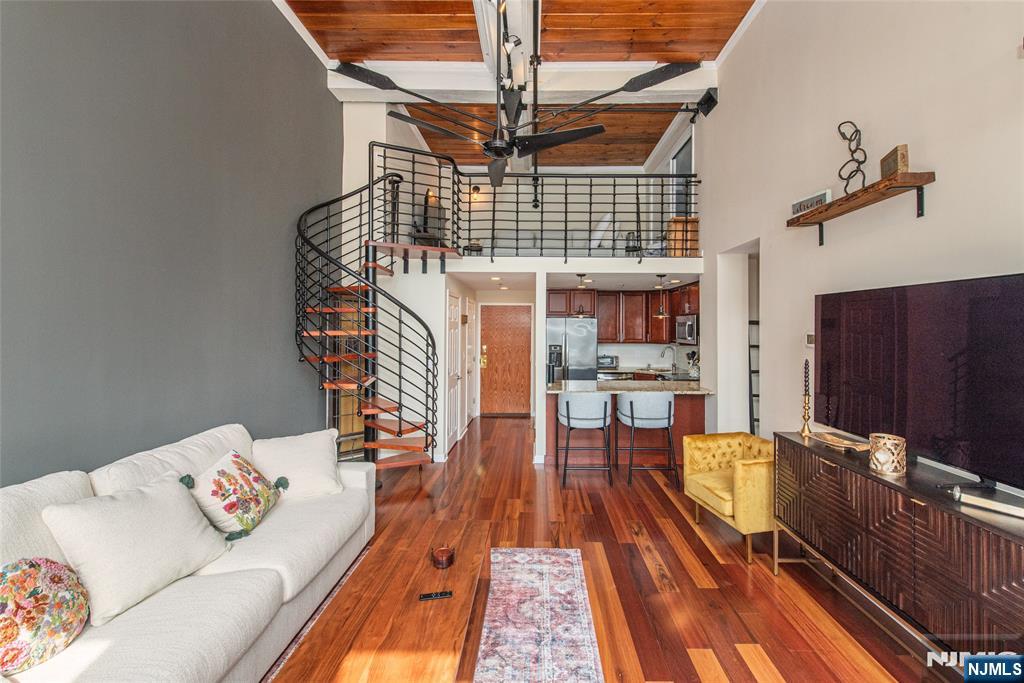 Volume, light, and character come together in a way that feels visually integrated and distinctly different from anything else at The Mill. This one bedroom residence is defined by its dramatic double-height living space, where soaring ceilings, exposed brick, and a full wall of oversized windows create a setting that is both open and striking. Natural light pours across rich hardwood floors, highlighting the scale of the room and the cohesive layout. Overhead, dual ceiling fans and track lighting accent the height of the space, while the brick backdrop and deep window sills bring in that signature Mill aesthetic. Positioned just off the living space, the kitchen is finished with warm wood cabinetry, granite countertops, stainless steel appliances, and a breakfast bar that offers seating. A custom full-sized loft sets this unit apart. Accessed by a eye-catching spiral staircase, the loft adds a completely separate level of living, ideal for a lounge, office, or creative space. Framed by industrial-style railings and set beneath wood-accented ceilings with exposed steel elements, it overlooks the main level. The bedroom continues the same clean aesthetic with hardwood flooring, oversized windows and complemented by a sliding barn-style closet system that adds design. This is a one-of-a-kind layout within The Mill, is rarely found.
