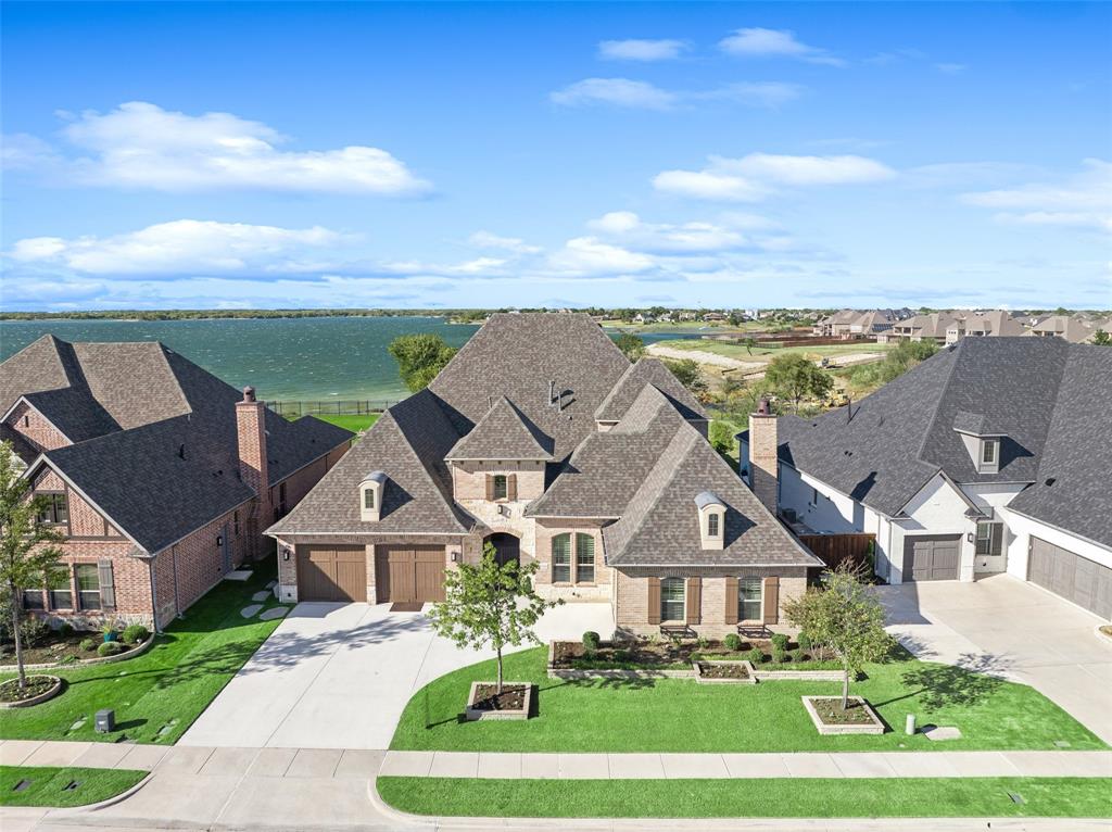 Wake up every day to the beauty of Lake Lewisville in this exceptional lakefront home, perfectly designed for relaxation and entertaining. From the moment you step onto the welcoming front porch, you’ll feel the charm and comfort this home offers. Inside, elegance meets warmth in the spacious dining room with a double-sided fireplace, wood-beamed ceiling, and custom built-ins—ideal for hosting family gatherings or dinner with friends. The open living area flows effortlessly through large sliding doors to the backyard, where lake views create a breathtaking backdrop to your daily life. The chef’s kitchen is both beautiful & functional, featuring a striking waterfall island, gas cooktop, double ovens, Miele coffee maker, and breakfast nook overlooking the water. The serene primary suite invites you to unwind with its tray ceiling, accent wall, and stunning lake views. The spa-like bath features a soaking tub, dual vanities, walk-in shower with seating and body sprays, and two spacious closets. Downstairs also offers a media room for movie nights, a private office, and two guest bedrooms with ensuite baths. Upstairs, a large game room with built-ins and beverage refrigerators opens to an expansive deck—perfect for taking in the sunset over the lake. A fourth bedroom & private bath provide extra space for family or guests. Step outside to your personal oasis. The covered outdoor living space features a wood ceiling, bar seating, full outdoor kitchen with BBQ & griddle, beverage fridge, cozy fireplace, and built-in hot tub with TV. Electric screens & ceiling heaters make this space perfect year-round. A lakeside firepit invites you to sit back, relax, & enjoy peaceful evenings under the stars. Residents of The Tribute enjoy resort-style amenities including pools, a fitness center, trails, clubhouse, and a new marina and lakeside park coming Summer 2026. Conveniently located near major highways, shopping, dining, & top-rated schools—this is lakeside living at its finest.