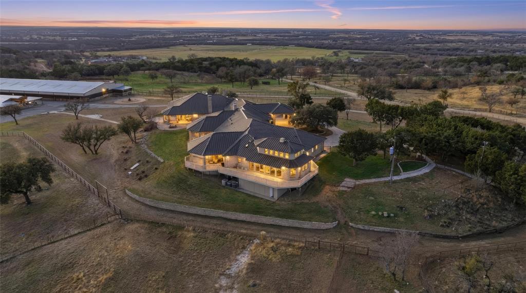 Set atop 29+ breathtaking acres, this gated estate commands one of the highest vantage points in Parker County, offering panoramic views and a rare blend of luxury living with premier equestrian amenities. Designed for grand-scale entertaining and refined ranch living, the impressive 10,438-sq ft custom residence showcases exceptional craftsmanship and expansive living areas. Walls of windows frame the rolling countryside, filling the home with natural light, showcasing incredible views from every room. Multiple gathering areas create the perfect setting for hosting family, guests, or large-scale events. The chef’s kitchen was designed for both beauty and performance, featuring high-end appliances, generous prep space, custom cabinetry, and fully equipped scullery kitchen. The primary suite offers a luxurious retreat, while additional guest accommodations provide comfort and privacy for visitors. Equestrian improvements rival elite training facilities. The covered arena allows for year-round riding and training, while the meticulously designed 19-stall show barn reflects exceptional attention to detail and functionality. Multiple spacious turnouts provide excellent horse management and flexibility, with ample implement parking and extensive equipment storage supporting the operation of a premier ranch property. The beautifully maintained acreage has an ideal blend of elevation, usable land, and scenic beauty, creating both an impressive setting and highly functional equestrian environment. A separate guest house offers additional flexibility for extended family, ranch staff, or visitors, complete with comfortable living quarters and privacy away from the main residence. From the gated entrance and scenic drive to the commanding hilltop setting, every aspect of this remarkable estate has been carefully curated to deliver unmatched lifestyle. Rarely does a property combine luxury, privacy, breathtaking views, and first-class equestrian infrastructure at this level.