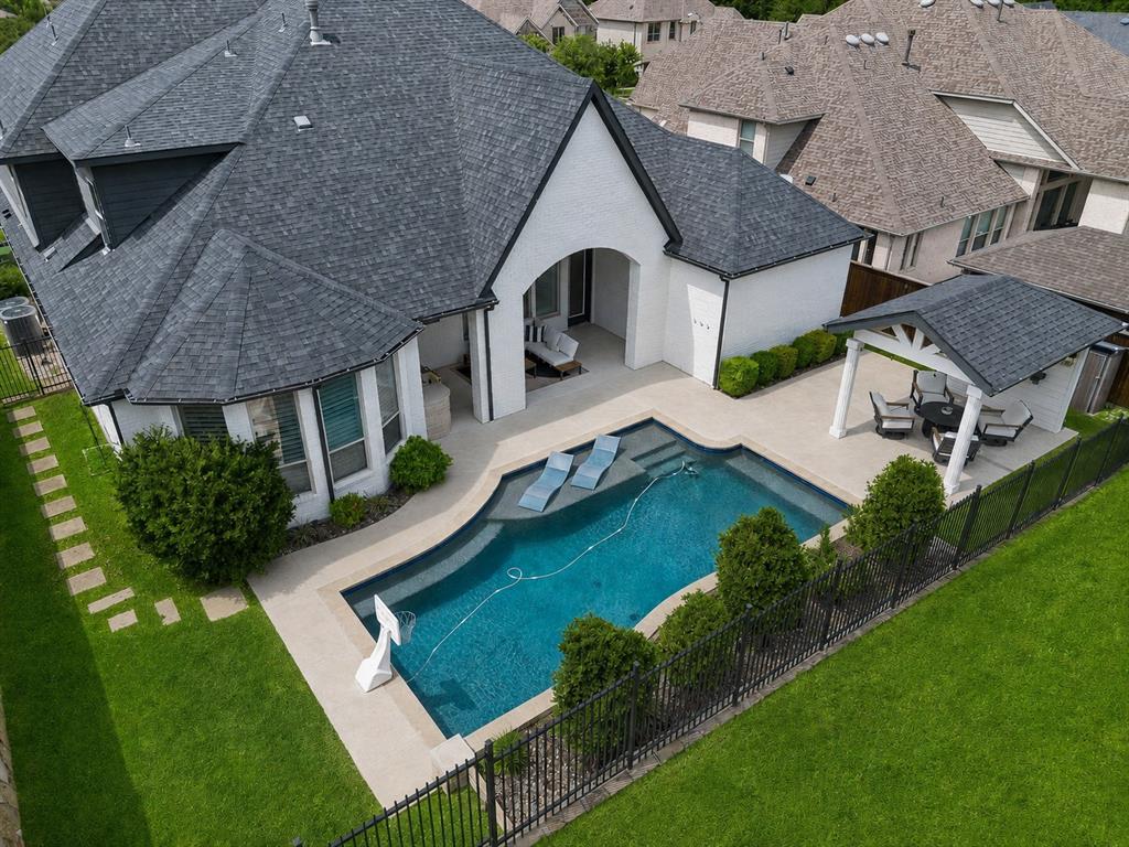 Positioned on a serene greenbelt lot in sought-after Whitley Place, this exceptional Highland Home combines luxury finishes, modern design, and resort-style outdoor living. Zoned to acclaimed Prosper ISD, including Walnut Grove High School and an on-site elementary school, this move-in ready residence offers an elevated lifestyle in one of Prosper’s most desirable communities. The modern farmhouse elevation features white painted brick and black accents, while engineered hardwood floors, custom shutters, soaring ceilings, and designer finishes create a sophisticated atmosphere throughout. The chef’s kitchen is a showpiece with an oversized quartz island, porcelain apron-front sink, marble backsplash, painted shaker cabinetry and premium GE Monogram appliances including a six-burner commercial-style range and double ovens. A custom wine station with wine refrigerator and display storage enhances both entertaining and everyday living. The dramatic great room showcases vaulted ceilings with beams and a gas fireplace. An oversized private study with French doors provides the ideal work-from-home space. The luxurious primary suite offers bay windows, a spa-inspired bath with dual vanities, glam area, quartz countertops, garden tub, oversized walk-in shower, and a custom closet with direct utility room access. A private guest suite, media room, oversized utility room, and mudroom complete the first floor. Upstairs features three additional bedrooms with walk-in closets and a spacious game room with wet bar, beverage refrigerator, and powder bath. Designed for year-round enjoyment, the backyard features a resort-style pool with tanning ledge and water feature, covered patio with built-in KitchenAid grill and kegerator, plus a separate patio pavilion with outdoor TV overlooking the greenbelt. Offering 5 bedrooms, 4 full baths, 2 half baths, a 3-car garage with epoxy floors and overhead storage, and exceptional indoor-outdoor living, this is luxury living at its finest.