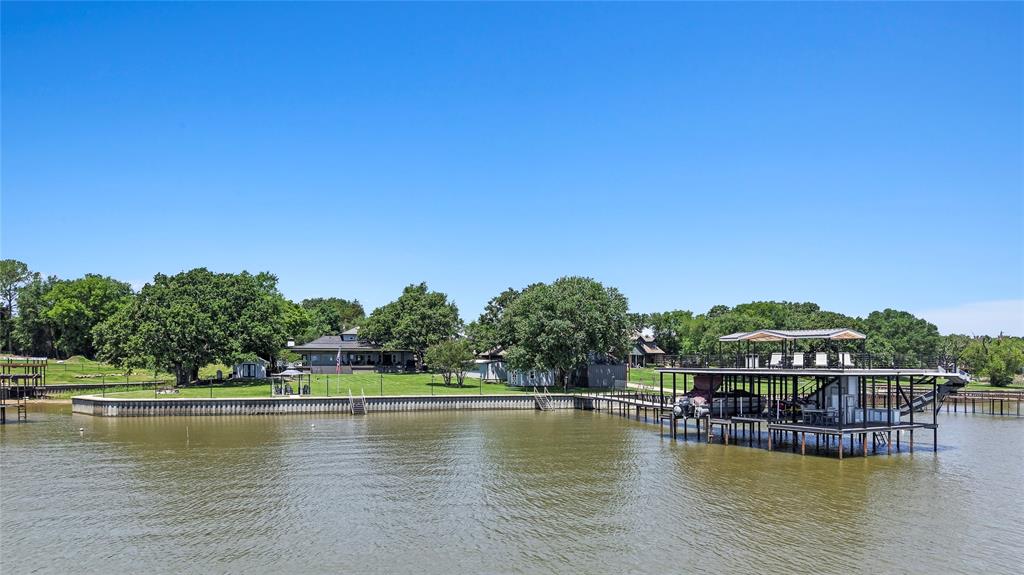 Approximately 528 feet of platted water frontage, a spectacular 1,960 sq ft composite entertaining deck with top-tier outdoor kitchen, new custom boathouse, and extensively remodeled home with guest quarters create an exceptional lake retreat. Offered fully furnished, this fully fenced property with gated entry and ample parking is ready to enjoy! The custom boathouse is a showpiece featuring composite decking, covered lounge areas, aluminum cool decking, signature waterslide, wet bar with full-size fridge and bar seating, one boat slip, and dual PWC lifts. Iron cable railing, tongue-and-groove wood ceilings, and a lighted 6-foot-wide walkway. A detached metal shop plus waterside storage building provide plenty of space for your lake gear. Outdoor living feels resort-like with quartz countertops, built-in Viking grill plus cooktop, Blackstone griddle, two Viking beverage fridges, storage, and bar seating. Multiple dining and lounge areas, mounted TV, heaters, fans, and mosquito misting system create an incredible year-round entertaining space, while the oversized yard offers room for future pool! Inside, the 3,957 sq ft main home offers 5 bedrooms and 2.5 baths. The expansive great room captures open water views and includes two living areas, two dining areas, and a chef’s kitchen equipped with all Viking appliances including an 8-burner gas range with double ovens, two add’tl wall ovens, built-in fridge, two dishwashers, ice maker, prep sink, pantry-style cabinetry, and large wine refrigerator. The waterside primary suite offers direct deck access updated bath with soaking tub, oversized walk-in shower, dual sink vanity plus makeup vanity, and walk-in closet. Upstairs loft space plus two guest bedrooms and full bath with dual sink vanity and walk-in shower. Bunk room adds a third living area plus custom bunks for large groups. Detached 456 sq ft guest quarters serves as a private 6th bedroom with full bath, open water views, fridge, microwave, and coffee station.