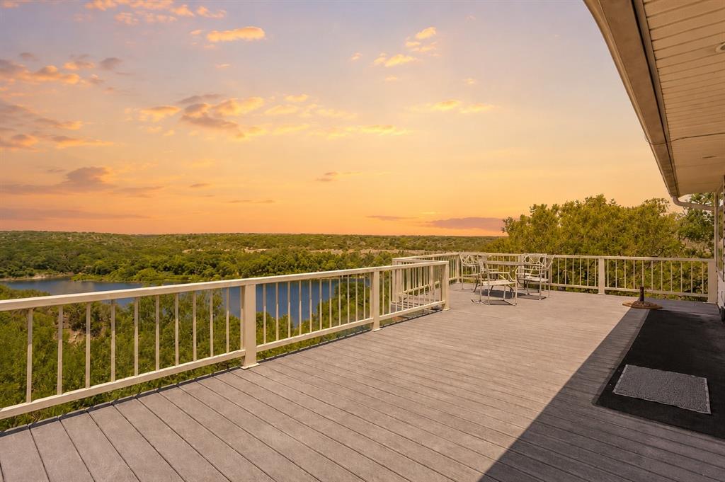 Perched above the sparkling waters of Lake Whitney, this one of a kind retreat blends elevated lake living with thoughtful design & unforgettable views. This stunning 4bd, 6ba home is crafted for both relaxation & entertaining, offering panoramic water vistas! Step inside & you’re drawn to the dramatic floor to ceiling windows that flood the home with natural light & perfectly frame the lake beyond. Rich natural woodwork throughout creates warmth & character, giving the home a timeless, lodge-inspired feel. The heart of the home opens to not one, but two full-length covered decks, ideal for morning coffee, sunset dinners, or simply soaking in the peaceful waterfront scenery. With direct access to U.S. Army Corps of Engineers land behind the property, your views are protected & your privacy enhanced. Designed with flexibility in mind, this spacious layout includes a large flex space that could be used as a game room, entertainment area or second living area, making it great for large families, hosting guests or multi-generational living. The upstairs primary suite is a true sanctuary, complete with its own private deck, a versatile flex space, perfect for an office, sitting area, or gym, & elevated views that make every morning feel like a getaway. Function meets convenience with a built in dumbwaiter servicing both levels, & a stair lift that can remain with the home for added accessibility if desired. Custom windows and coverings add both style & efficiency throughout. Added bonuses: Durable metal roof paired with fully paid-off solar panels for long-term energy savings, private shared well system & access to a community boat dock & ramp for effortless days on the water. Whether you’re searching for a full-time residence, weekend escape, or investment opportunity, this lakefront gem offers the perfect combination of comfort, efficiency, & breathtaking natural beauty. Come experience lake living at its finest, where every day feels like a vacation.
