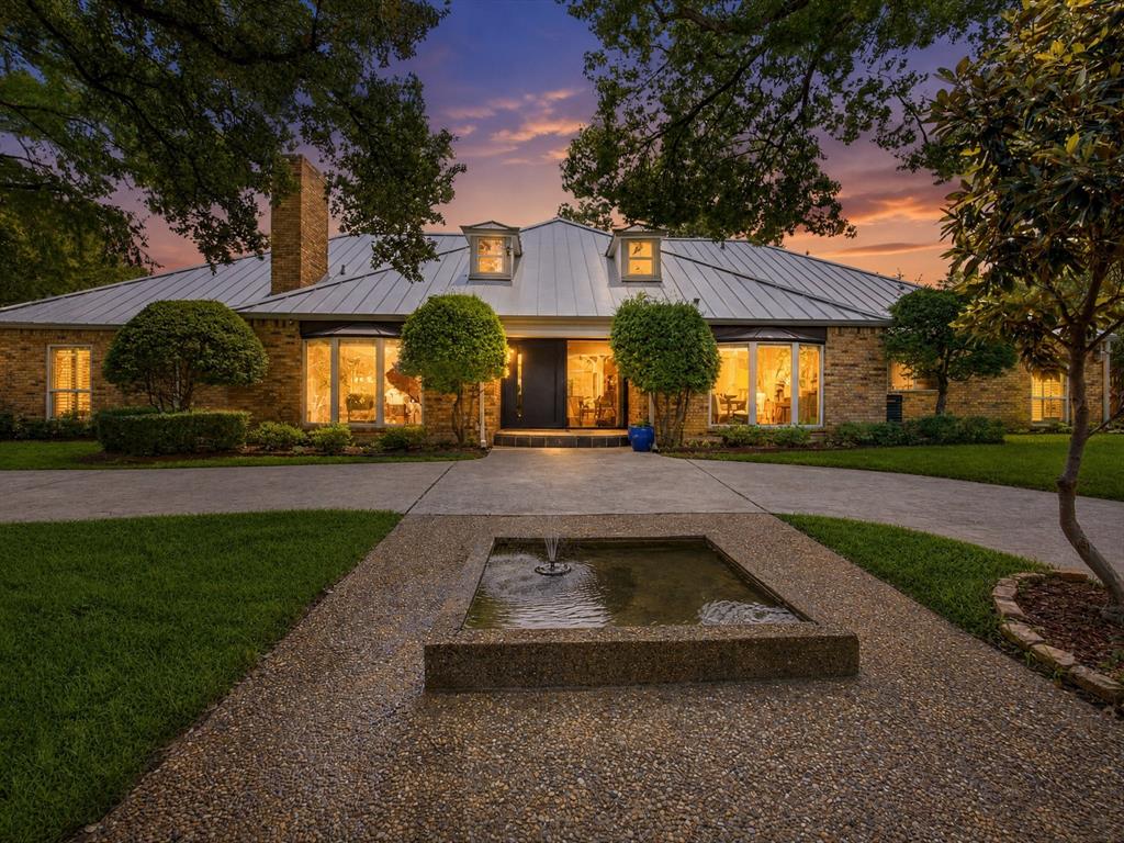 A private, gated retreat in Dallas, 6160 Spring Valley Road offers a secluded garden setting near Uptown, Addison, and the Galleria area. Heavy iron-and-wood gates open to a fountain-centered approach and expansive manicured grounds shaded by mature trees. White magnolias, fragrant lilacs, glossy evergreens, and crepe myrtles in blush, crimson, ivory, and coral provide seasonal color. Oversized picture windows frame the greenery and fill the home with natural light, while sliding doors open to the backyard for seamless indoor-outdoor living. The main living room features soaring vaulted ceilings, exposed beams, and an elegant fireplace. A separate game room includes built-in shelving, another fireplace, backyard access, and a nearby guest half bath. Multiple living and dining areas offer flexibility for everyday living, recreation, and entertaining. The kitchen features marble countertops, abundant cabinetry, a charming herb window, stainless gas cooktop, double ovens, microwave, and paneled Electrolux refrigerator, along with generous preparation and seating space. The first-floor primary suite opens directly to the yard and terrace. Its spa-style bath includes a soaking tub, separate glass-enclosed shower, and dual walk-in closets. Outdoor amenities include multiple terraces, a spacious balcony with double-door access, a raised entertaining deck, outdoor kitchen, and steam room. The broad lawn offers room for a generous pool, and Jacuzzi cabling is already installed. An extra built-in condenser provides supplemental cooling. Two spacious attics beneath the standing-seam metal roof offer expansion potential for multiple additional rooms and another restroom. Additional details include hardwood flooring, decorative molding, statement and recessed lighting, multiple fireplaces, and exposed-brick accents. Nearby destinations include Addison, Galleria Dallas, Uptown, Vitruvian Park, Winnwood Park, and White Rock Creek Trail.