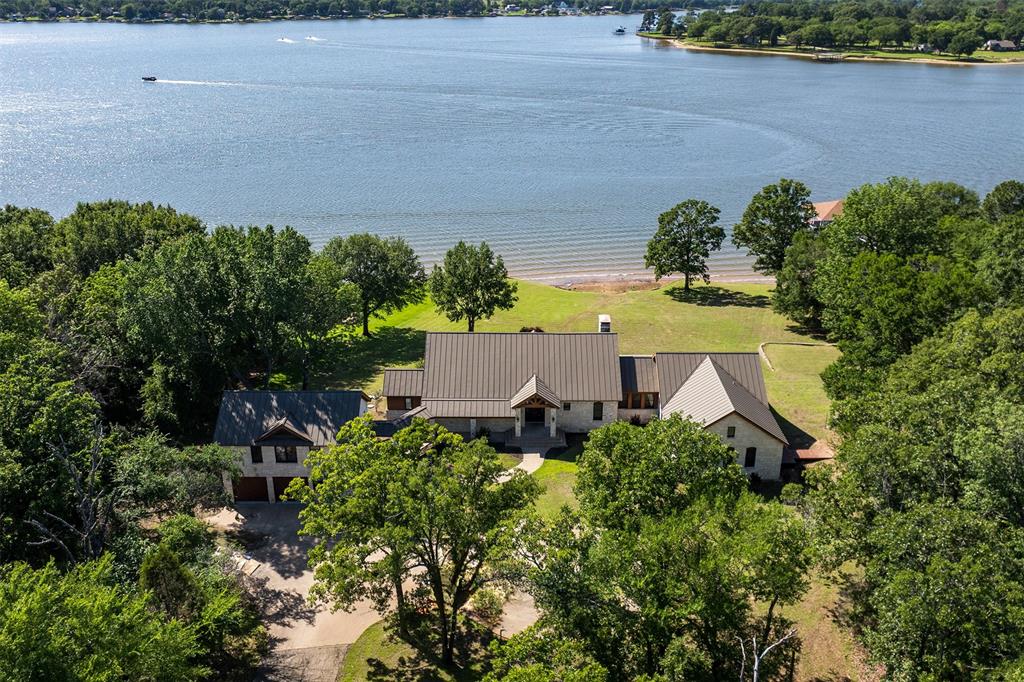 Experience the ultimate in lakeside seclusion on this heavily wooded 5-acre estate, boasting 299 feet of platted shoreline and breathtaking, truly panoramic wide-open water views. Located within an exclusive gated community, this rare property offers unmatched privacy and timeless Hill Country style. The sprawling single-level main house features an effortless open floor plan anchored by a majestic stone fireplace. Designed for entertaining and multi-generational living, the home includes 5 spacious bedrooms and 5.5 baths, a small office, a wet bar, and a large game room. The massive covered back patio and extended deck span the entire width of the home, leading you toward the water’s edge. The ultimate lake lifestyle awaits at the oversized boathouse, featuring two large boat slips, a twin PWC lift, and expansive decking perfect for lakeside entertaining. Above the garage, a separate guest quarters with a kitchen and bath provides a private retreat for visitors. This is more than a home; it’s a legacy waterfront sanctuary.