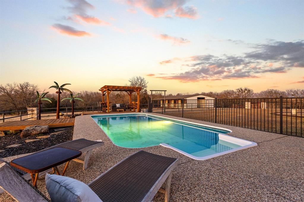 Experience the ultimate Texas sunset from this private 18+ acre oasis. Whether you’re soaking in the hilltop pool, unwinding on the expansive patio, gathered around the firepit, or relaxing in the hot tub, you’ll enjoy evening panoramic vistas that put you front-row to the best show in North Texas.
Bathed in natural light & curated with impeccable style, this breathtaking home offers sweeping views that create the perfect backdrop for everyday living. Designed for seamless indoor-outdoor flow, the open-concept layout centers around a striking stone fireplace, creating a warm, inviting atmosphere that opens effortlessly to multiple outdoor entertainment spaces. The kitchen offers endless counter space & large island with seating making hosting a breeze, flowing into a dramatic dining area with soaring ceilings & a picture window frames stunning pool & ranch views. The home was originally designed as a three-bedroom & has been thoughtfully converted to a spacious two-bedroom layout, enhancing the living areas while maintaining comfort & functionality. Two generously sized bedrooms & two full baths provide quiet retreats to end the day. Detached flex space offers excellent versatility & would make an ideal home office, gym, studio, or creative workspace. The courtyard extends across the back of the home, leading to a hot tub, sauna, & fenced pool area—perfect for relaxation & entertaining. Outdoor amenities include a covered RV area with water and a 50-amp hookup & designed to be easily converted into a full-size pickleball court, an impressive six-stall metal barn with wash rack, tack room, electricity, & water, plus a nearby corral with water access. Pastures are fenced & cross-fenced, complemented by a stock tank & mature trees. Currently ag-exempt, this ranch is comfortable, functional & ready for your livestock. This isn’t just a ranch—it’s a lifestyle.