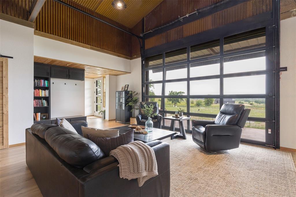 Nestled on 10 picturesque acres just north of Decatur, this architecturally striking residence is a masterclass in thoughtful design, craftsmanship, and modern country living. Created from two shipping containers and meticulously engineered to embrace the surrounding landscape, the home captures sweeping views of rolling green hills while offering a seamless balance of industrial character and refined comfort. Inside, soaring 18-foot ceilings, warm custom woodwork, luxury vinyl flooring, and expansive sight lines create an atmosphere that feels both dramatic and inviting. The living room was designed to blur the line between indoors and out, featuring a motorized glass roll-up door with retractable screen that opens the home entirely to the countryside beyond. A large statement ceiling fan anchors the space while maintaining the home’s open, airy feel. The kitchen showcases soapstone countertops, custom cabinetry, and a lift-up garage-style window that connects directly to the expansive covered porch with fireplace, creating an effortless flow for entertaining. The floor plan includes two bedrooms, two full bathrooms, an office or gym space, walk-in pantry, and a spacious laundry room with utility sink and built-in dog kennels. The primary suite features dual vanities, a soaking tub, and separate walk-in shower. Additional highlights include a putting green, native low-maintenance landscaping, gravel drive, metal hog-wire fencing, large storage building with well house, and a 2,500-gallon water holding tank with DAB pump system. Complementing the residence is an exceptional 39x40 insulated metal shop with closed-cell spray foam insulation, multiple roll-up doors, and flexible workspace or storage potential. Split-unit systems in the bedrooms and central HVAC in the main living areas provide year-round comfort and efficiency. A carefully composed retreat where craftsmanship, architecture, and the beauty of the North Texas landscape come together beautifully.