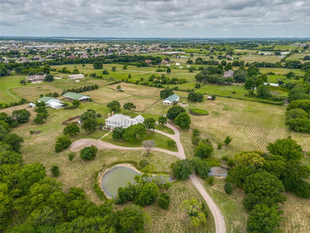 Stunning gated Southern Colonial estate set on 21.6± private acres in Denton County, nestled within the sought-after Prairie Acres community and ideally located between Little Elm, Providence Village, Cross Roads, and Aubrey. Set nearly one-eighth mile off the road, this impeccably maintained property offers exceptional privacy, timeless architecture, and refined rural living. Meticulously designed to reflect classic antebellum colonial styling, the home features three spacious bedrooms, a dedicated office or optional fourth bedroom, and three and a half baths. Three beautiful fireplaces anchor the thoughtfully designed interior, while sweeping views from every window, door, porch, and balcony bring the surrounding landscape indoors. The light-filled interior offers an inviting chef’s kitchen with breakfast area, sizable pantry, and dumbwaiter, along with two elegant living areas and a formal dining room ideal for entertaining. Generous storage, an expansive mudroom, and a well-appointed laundry room add everyday functionality. The home’s classic appeal is further enhanced by a standing-seam metal roof and fenced front and backyard spaces. The agricultural and wildlife exemption supports livestock, hay production, and abundant wildlife, with regular sightings of whitetail deer and waterfowl drawn to the stocked pond and seasonal creek. Native pecan, oak, and elm trees create a picturesque, park-like setting. Equestrian and hobby amenities include an impressive eight-stall barn with tack room, stocks, and ample storage, four loafing sheds, a chicken house, and designated garden area. Additional improvements include a 50’ x 50’ shop and a 40’ x 25’ workshop. The property is fully fenced and cross-fenced for versatility and ease of use. A rare opportunity offering peaceful country privacy with convenient access to city amenities — truly an exceptional estate for those seeking elegance, functionality, and space to live fully.