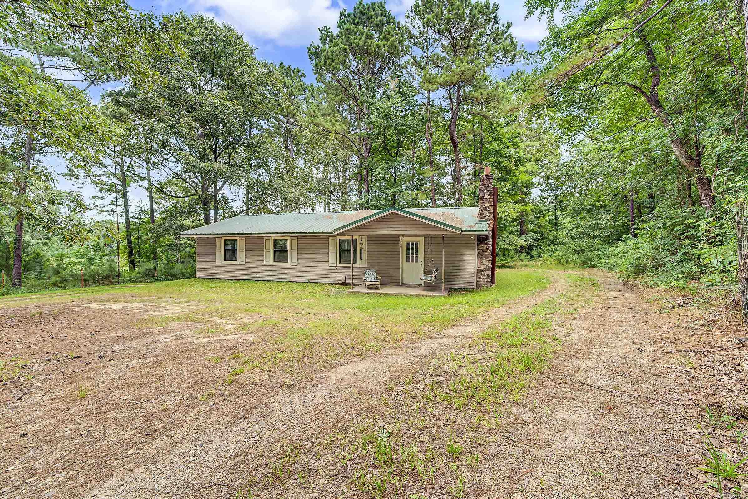 Tucked away on 1.5 +/- wooded acres just outside De Queen, this well-maintained 2-bedroom, 1.5-bath home offers the perfect blend of privacy and comfort. The open-concept living, dining, and kitchen area creates an inviting space for gathering, while the beautiful stone hearth with a wood-burning stove adds warmth and rustic charm. The spacious primary suite features a large ensuite bath complete with a soaking tub, separate shower, and generous vanity space. A large utility room with a half bath provides additional storage and everyday convenience. Outside, enjoy the peaceful setting from the covered front porch, mature pine trees, a circular gravel drive, carport, and an additional storage building with covered lean-to. The home also features a durable metal roof, central heat and air, public water with a private well, and a level lot offering plenty of room to enjoy the outdoors. If you’re looking for quiet country living with modern conveniences just minutes from town, this property is ready to welcome you home.