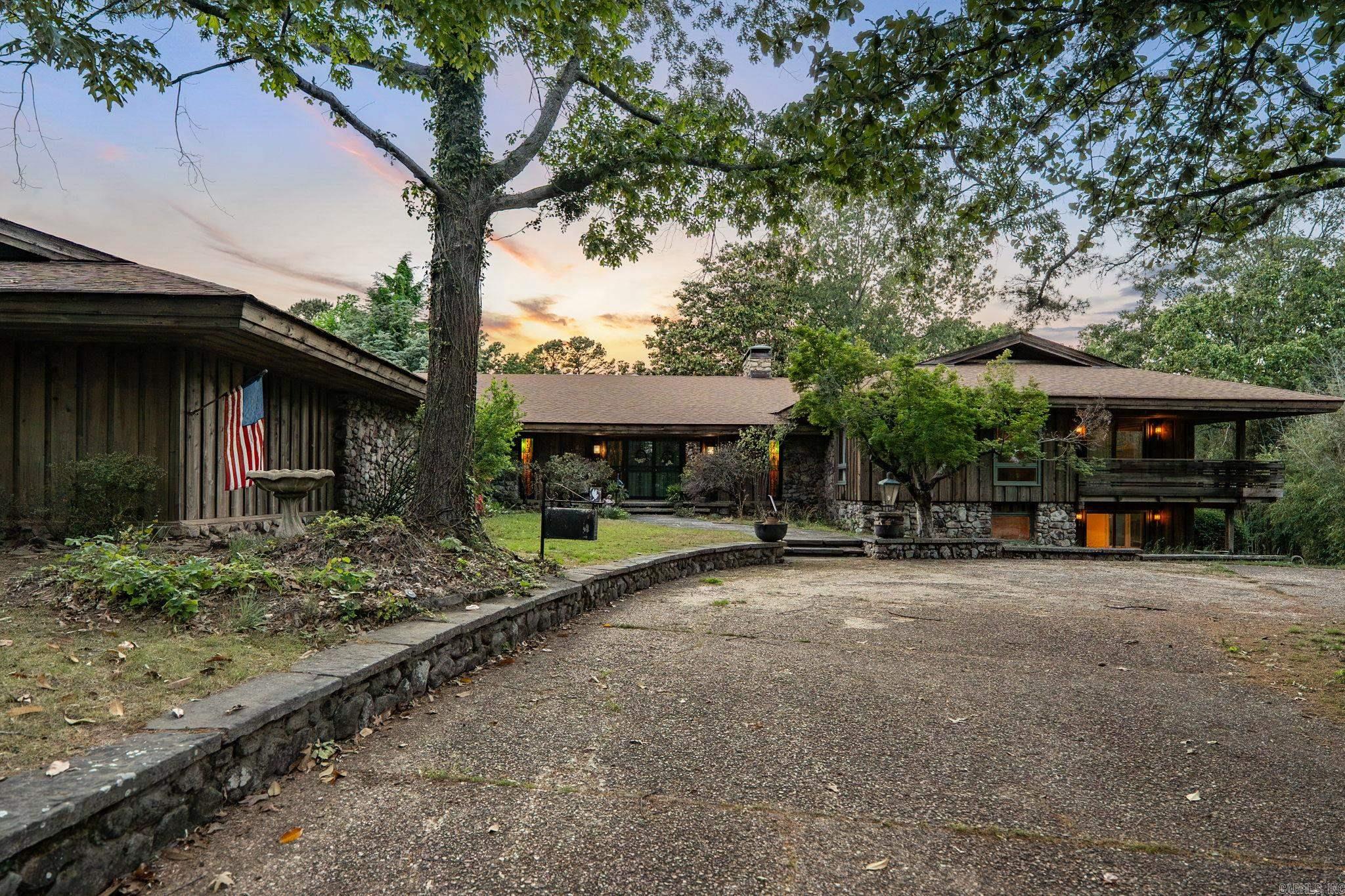 Distinctive mid-century modern home set on 7.7 acres with a gated drive, built under advisement of noted Arkansas architect E. Fay Jones, offering 5 bedrooms and 5.5 baths. The rock and frame exterior welcomes you into a center hall with slate floors and rock flanked circular staircase to lower level. A formal living room with fireplace, built-ins, and sliding doors opens to a covered patio and flows to the formal dining room. The spacious Den/family room features wood-beamed ceilings, an impressive rock fireplace, and outdoor access. Oversized kitchen includes bar area, pantry, double ovens, a large central island with grill and dual cooktops, plus an informal dining area. Laundry and office/craft room as well. The primary suite is expansive with a dual layout bath, walk-in shower, tub and generous closet space. Two additional bedrooms share a bath and deck access. Lower level leads to a bonus game room with wet bar, fireplace, and outside access, along with two bedrooms, each with a private bath. Covered porches and decks, an outbuilding/potting shed, and a large garage with storage complete the property. Truly one of a kind.  Come see!