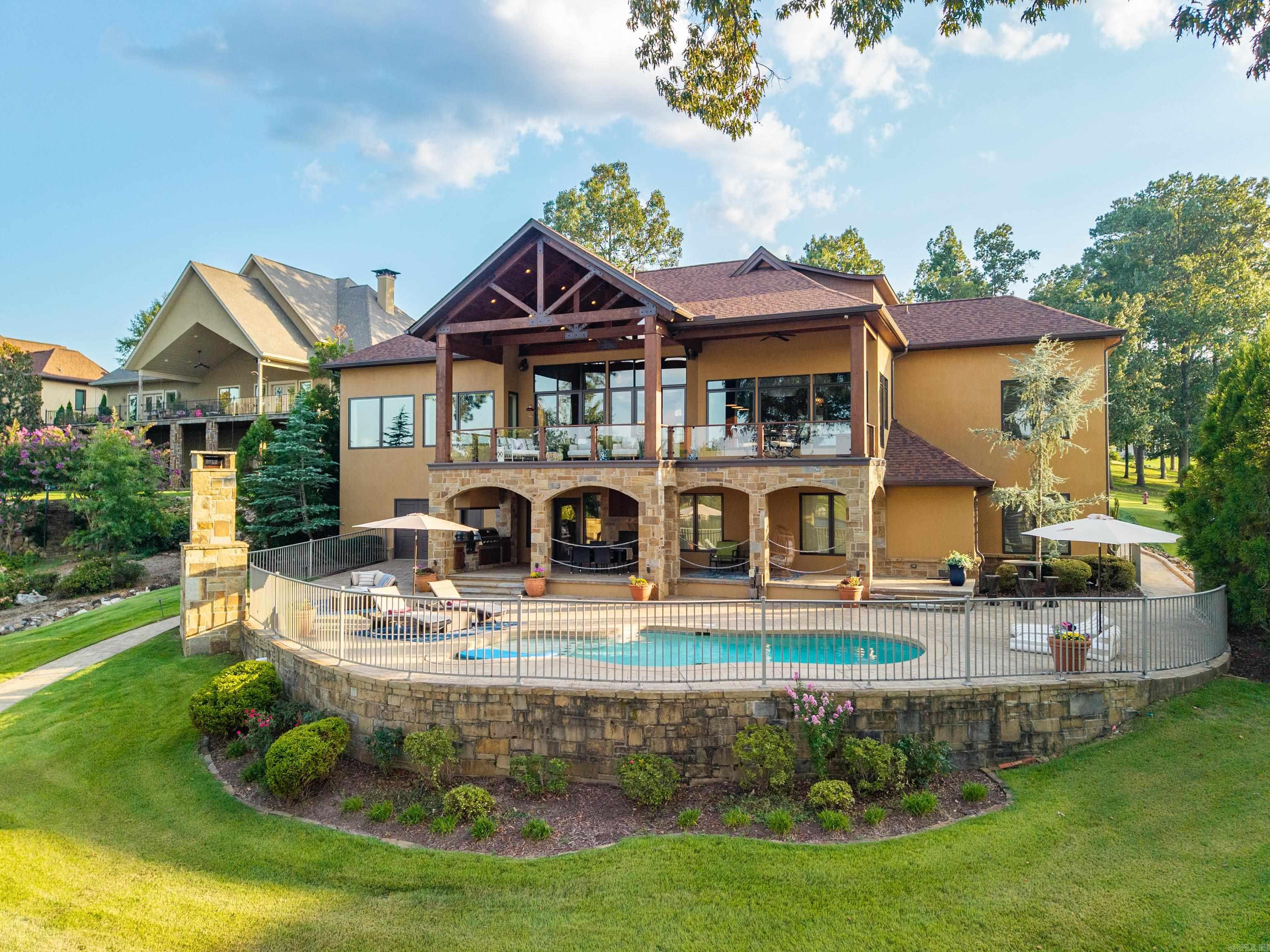 Welcome to the marriage of exquisite craftsmanship and contemporary elegance. Nestled in prestigious Oak Shores Estates on the serene southeastern waters of Lake Hamilton, this grand villa impresses from the start with Arkansas-quarried stone, timber gable trusses, and vaulted tongue-and-groove ceilings. Inside, massive cedar beams, custom woodwork, and floor-to-ceiling windows frame breathtaking lake views. A glass-railed veranda extends the vistas, while open-concept living and dining areas flow seamlessly from the gourmet kitchen. This exceptional home offers 5 bedrooms, 5.5 baths, three living areas, a private office, craft room, and a lower-level guest kitchen with bar—perfect for entertaining. The primary suite features electronic shades that reveal stunning lake sunrises each morning. Outdoors, enjoy a saltwater pool, full kitchen, expansive patio, and private covered dock for effortless lake access. Blending timeless elegance with modern convenience, this villa offers an unparalleled resort-style lifestyle on Lake Hamilton.