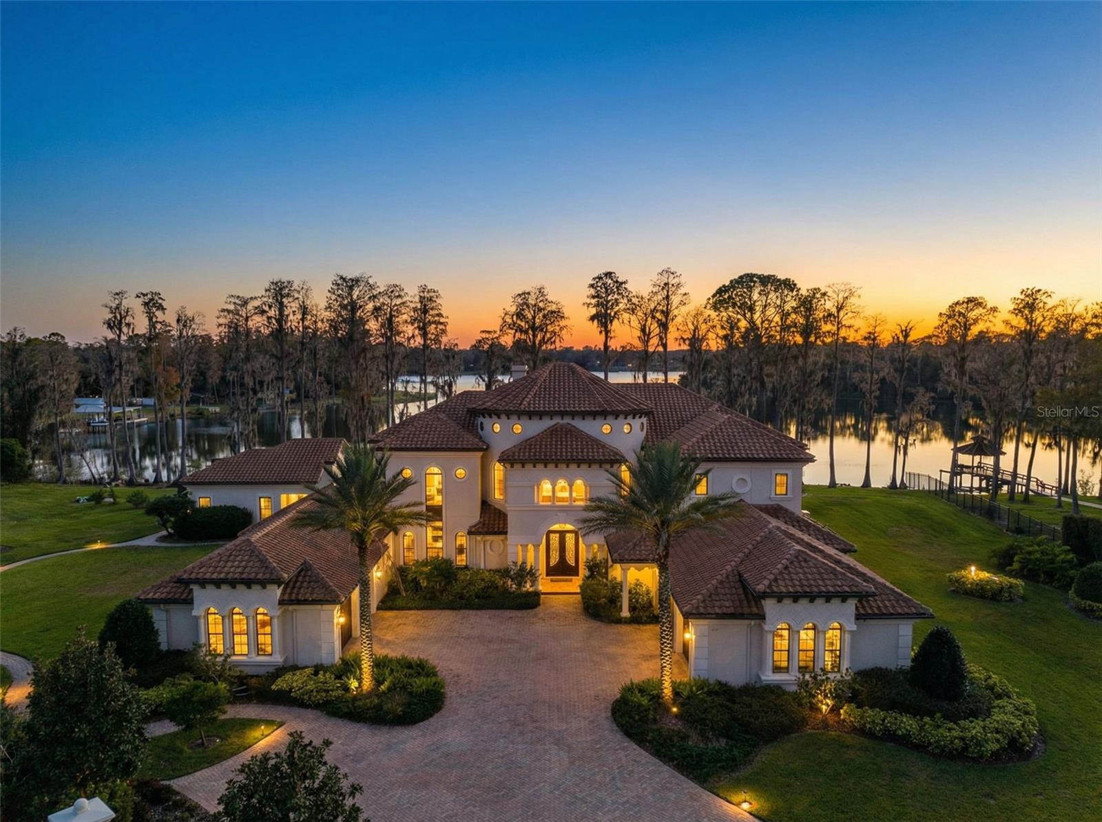 Introducing 17124 Crawley Road, a gated Mediterranean lakefront estate where refined architecture meets resort-caliber living. Set on nearly six acres along the shores of Rainbow Lake, this custom Arthur Rutenberg residence showcases a signature custom floor plan not available to the public, offering six bedrooms, seven full baths, two half baths, and four garage spaces within a double gated private retreat designed for both grand entertaining and everyday luxury. Built in 2017 and encompassing approximately 10,545 total square feet including a detached guest cabana (596 Sq. Ft), the home opens with a dramatic double-door entry and soaring foyer crowned by a sweeping dual staircase. Expansive windows frame tranquil water views and fill the interiors with natural light. The formal living room impresses with 24-foot ceilings, a gas fireplace, and panoramic pool-to-lake vistas, while the family room’s pocketing sliders create seamless indoor-outdoor living. At the heart of the home, the designer kitchen blends statement style with function, featuring a Cianitus granite island, Cambria Menai quartz surfaces, gas range, wall oven, pot filler, designer lighting, and an elegant butler’s pantry with wine refrigeration. An oversized formal dining room offers a stunning setting for gatherings both intimate and grand. The main-level primary suite is a private sanctuary complete with fireplace, dual custom walk-in closets, and a spa-inspired bath with freestanding soaking tub and oversized shower. Additional ensuite bedrooms, a fitness room, and a soaring study provide flexibility, while upstairs offers oversized guest suites, a sunken game room, bar, and lakefront balcony with pocket sliders elevating the entertaining experience. Designed by Ryan Hughes Design, the outdoor environment rivals a private resort with a heated saltwater pool and spa, covered terrace, full outdoor kitchen, fire pit, and lush landscaping. Paver pathways lead to a private dock installed in 2021, offering direct lake access for boating, sunset cruises, or fishing. A separate one bedroom, one bathroom guest cabana provides ideal accommodations for visitors or multigenerational living. Additional highlights include two two-car garages, EV charging station, dual laundry rooms, custom closets throughout, hurricane-rated windows and sliders (with one shuttered area), surround sound, fruit trees, and intricate crown and tray ceilings. Located in Odessa, one of Tampa Bay’s most coveted enclaves known for estate homesites, natural beauty, and privacy, this property offers a rare blend of lakefront living with convenient access to top schools, dining, shopping, major highways, and Tampa International Airport.