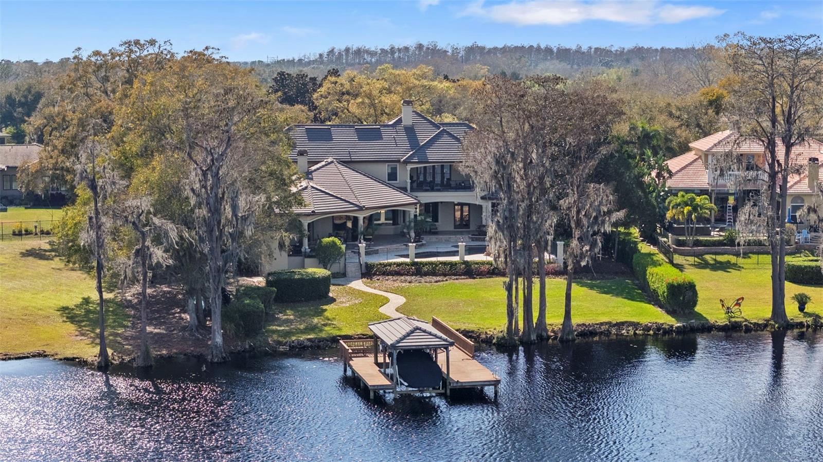 Experience refined lakefront living in this one-of-a-kind custom estate on ski-size Lake Pretty, tucked away at the end of a quiet cul-de-sac inside the gated Northbridge community. With an impressive 10,614 square feet under roof, this estate offers an extraordinary amount of covered space rarely found on lakefront properties.    Located in a non-flood and non-evacuation zone, this exceptional residence captures sweeping panoramic water views from nearly every room. A picturesque drive through a tree-lined private drive leads to a circular driveway and impressive motor court with two oversized garages—one attached and one detached with space for two vehicles plus a golf cart. A tranquil front entry with a cascading waterfall creates a striking first impression. Inside, the home has been dramatically lightened and brightened with fresh interior paint, brand-new carpet upstairs, and newly installed automatic blinds, creating a crisp aesthetic that complements the natural light and stunning lake views. Grand windows and French doors frame the water, while the elegant foyer showcases a dramatic spiral staircase, a lakeside sitting area, and a formal dining room flowing into the heart of the home. The chef’s kitchen features a 48-inch six-burner gas range, 72-inch built-in refrigerator, dual dishwashers (one elevated), an expansive island, and a walk-in pantry. A secondary staircase adds functionality and disappearing 10-foot sliders open the family room to the lanai for seamless indoor-outdoor living. Just off the kitchen, a dining nook with a fireplace and a bonus room with a built-in bar and pool table space make entertaining effortless. The primary suite is situated on the first floor, offering lakefront views from soaring windows with automatic blinds, French doors to the lanai, a custom walk-in closet, and a spa-inspired bathroom with a built-in sauna, soaking tub, dual vanities, bidet, and oversized walk-in shower with rain feature and full body shower heads. Additional first-floor highlights include a private office with lake views, a fitness room/safe room, bonus space, and guest baths. Upstairs, as you enter from the grand stairwell, you are greeted by the loft and signature balcony giving you even more amazing views for that morning coffee capturing the sunrise over the lake. A secondary laundry closet, a private in-law or nanny suite includes a wet bar, built-in desk, walk-in closet, en suite bath, and direct balcony access. Another bedroom has its own en suite bath and walk-in closet, while two additional large bedrooms share a Jack-and-Jill bath. The expansive elevated outdoor living area features a fireplace, a full outdoor kitchen, a pool bath, and generous covered areas for dining and lounging. The pool and spa have been upgraded with new automation, a variable-speed pump, and two LED lights. The resort-style pool and spa showcase an overflow waterfall, while the private dock and boat lift with a lakeside shower complete this exceptional property. Don’t miss this opportunity to own one of Tampa’s most stunning ski lake estates.