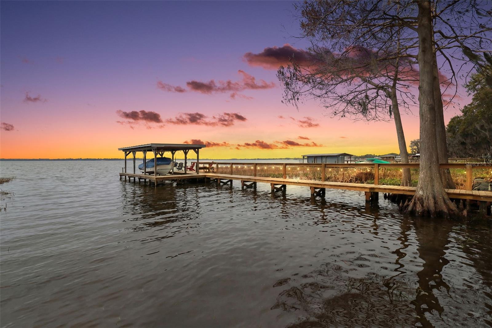 LAKEFRONT PARADISE ON LAKE DORA – THE HARRIS CHAIN OF LAKES. Imagine sun-drenched days, gentle lake breezes, swaying palms, and vibrant sunsets reflecting across the sparkling waters of Lake Dora and the coveted Harris Chain of Lakes. This is your opportunity to claim a private slice of tropical lakefront bliss — complete with a recently updated dock with covered boat slip. Start your mornings with coffee on the dock as the water softly laps the shoreline, and wind down your evenings under painted skies as nature provides the perfect soundtrack. Launch a kayak or cruise out on your boat to explore Lake Dora’s hidden coves, winding canals, and interconnected lakes — offering endless adventure, dining destinations, waterfront festivals, and year-round recreation. Set back from the road on a lush .69-acre homesite framed by grand cypress trees, towering pines, and ornamental palms, this lovingly maintained home welcomes you with warmth and tranquility. Major updates include A/C (2025), septic system (2025) and roof (2020) for peace of mind. A long driveway leads to an expanded parking pad and oversized two-car garage. A paved path guides you to the inviting front porch, and once inside, you’re greeted by expansive floor-to-ceiling windows showcasing breathtaking panoramic lake views. Natural sunlight fills the spacious interior, creating a bright and uplifting atmosphere. Designed for effortless entertaining, the flexible floor plan features attractive, low-maintenance tile and terrazzo flooring throughout. The front sitting room, kitchen, and combined living/dining areas all capture stunning water views and provide access to the beautiful lanai, where surrounding slider windows maximize breezes and relaxation. The open kitchen is both functional and inviting, offering wood cabinetry, stainless steel appliances, and an oversized walk-in pantry, which connects the spacious laundry room, complete with sink and additional storage. The split-bedroom layout ensures privacy for family and guests alike. The primary suite is a peaceful retreat where you’ll wake each morning to serene lake views. The en-suite bath features a quartz vanity, tiled tub/shower, and walk-in closet. Two additional bedrooms share a full hallway bath with a quartz vanity and shower — one guest room even enjoys beautiful lake views. Step outside to discover multiple backyard patios ideal for summer BBQs and gatherings. With generous space to design your dream outdoor oasis — think decks, firepit, or even a hot tub — the possibilities are endless. And of course, the true magic awaits at the water’s edge. Wander down to the dock, feel the breeze, and let the sparkling lake restore your spirit. Located near parks, popular restaurants, shopping, and all that vibrant Tavares — America’s Seaplane City — has to offer, including lively waterfront festivals and community events. Come live your best Florida lake life in waterfront paradise.