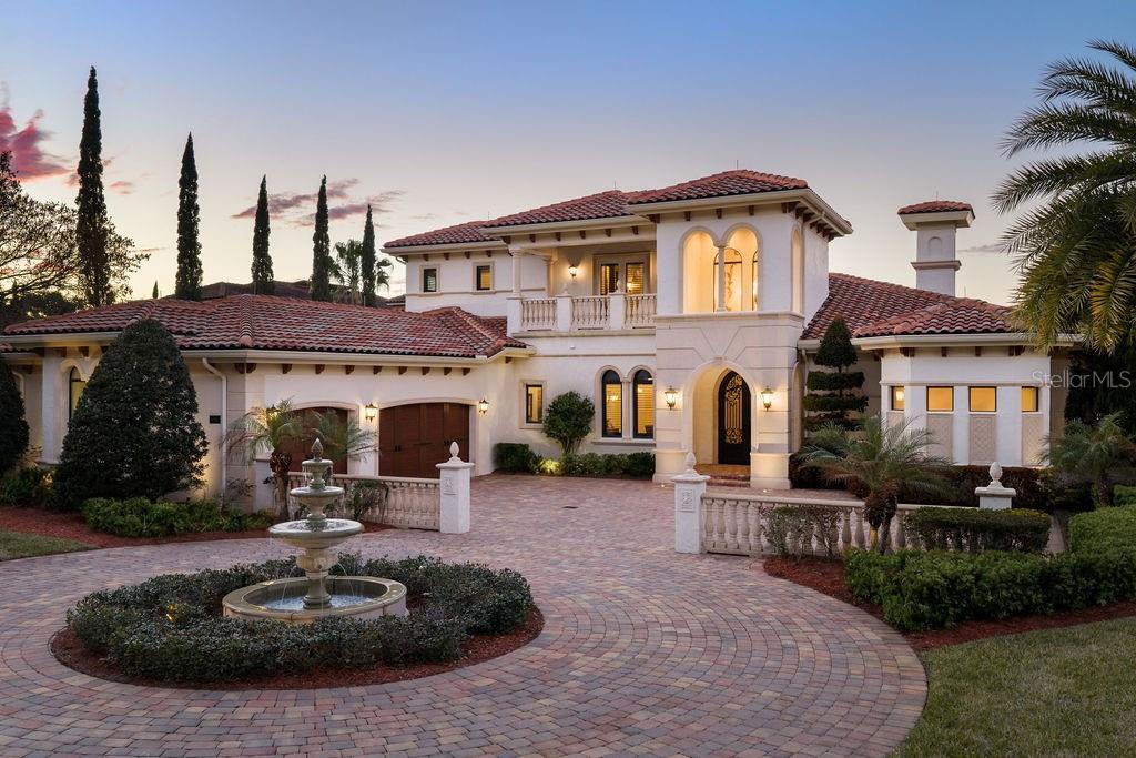 Experience over an acre of elevated direct sunset lakefront luxury living on the naturally spring-fed waters of John’s Lake. Offered publicly for the first time, this fully custom estate represents a rare convergence of beauty, privacy and enduring design. Nearly 5,000 square feet of interior living space unfold effortlessly into an additional 1,000 square feet of exceptionally well-appointed enclosed outdoor living, creating seamless indoor to outdoor living. Positioned within the private gated enclave of Deer Island and presented in impeccable condition since its 2015 construction, the home reflects meticulous care and pride of ownership. Thoughtfully designed for a life well lived by owners and guests alike, every detail was executed with intention. Arrival is elegant and memorable, welcomed by a central fountain and expansive motor court leading to an oversized three-car garage, while two bedrooms on the main level, including the primary and guest suite, provide comfort, privacy and effortless livability for generations. Designed for entertaining, walls of fully retractable sliding glass doors dissolve the boundary between indoors and out, allowing the living and great rooms to open to the terrace beyond. The covered outdoor lounge, finished with a wood-paneled ceiling, gas fireplace and fully equipped outdoor kitchen, extends daily living outdoors. The infinity pool and spa, framed by gas fire bowls, merge seamlessly with the lake view itself, while a dedicated pool access bath enhances convenience. Privately positioned to capture sweeping lake views, the primary suite opens directly to the lanai and features dual walk-in closets. Its bath is anchored by a freestanding copper soaking tub, hammered copper sinks set within custom vanities, and a full stone walk-in shower with multiple showerheads and built-in bench. The kitchen features custom wood cabinetry, oversized island, induction cooktop, separate built-in refrigerator and freezer, professional-grade appliances, and walk in pantry. Architectural details include travertine flooring, coffered and tray ceilings, refined millwork and wrought iron staircase. All closets feature custom built-ins. The second-floor media room may also serve as a spacious fifth bedroom with private balcony overlooking the lake. Beyond the terrace, tiered gardens descend toward the shoreline, where Four Seasons marble statuary is thoughtfully placed throughout the grounds, like a work of art set within the landscape. A private putting green, fire pit and manicured lawn enhance the setting. The composite dock includes dual lifts for boat and jet ski, water and electric service, and upper level sun deck positioned for sunset views. John’s Lake spans over 2,500 acres and is known for exceptional clarity, ideal for skiing, boating, water sports and fishing. Deer Island offers gated entry with waterside tennis, pickleball and basketball courts. The home is serviced by a private well. Less than one mile from the Florida Turnpike and 25 minutes to downtown Orlando. Contact for your private viewing.