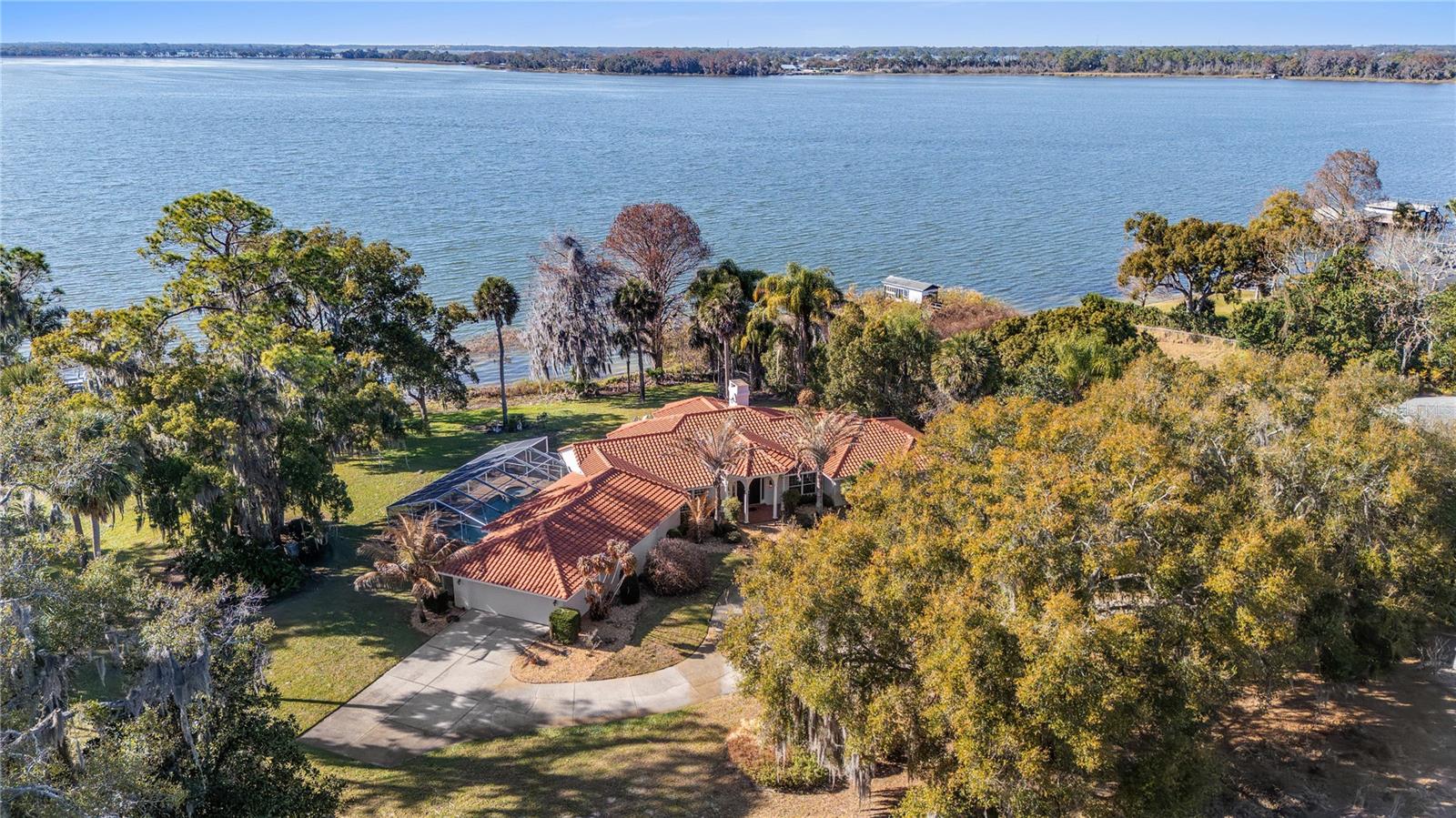 Envision the arrival of your private lakefront estate on the storied shores of Lake Beauclair with direct access to Lake Dora, the crown jewel of the Harris Chain of Lakes, a vast and celebrated waterway offering endless miles of lock and river cruising for days defined by leisure and adventure. Life on the lake unfolds at a gentler, more inspired pace as mornings begin with coffee on the dock while mist rises from the water, afternoons invite boating, paddleboarding and fishing along scenic shorelines, and evenings conclude with unforgettable sunset cruises beneath painted skies. Just moments away, the charm of Mount Dora enriches the lifestyle even further. Known for its storybook streets, vibrant arts scene and year-round calendar of festivals, residents enjoy boutique shopping, waterfront dining, antique markets, boat shows, food and wine celebrations, and lively community gatherings that make every season feel like an occasion. Across a remarkable eight-acre parcel, 6136 Dora Drive commands hundreds of feet of pristine shoreline, ideally oriented to the west for breathtaking sunset vistas that paint the water in gold each evening. This extraordinary canvas invites limitless possibilities such as design a grand equestrian compound, cultivate a private orchard, create an inspired guest residence with an expansive collector’s garage or explore opportunity to thoughtfully subdivide. Within a canopy of mature trees, a sprawling single-story residence spans approximately 4,000 square feet under roof, offering three bedrooms, two and a half baths, walls of windows, a screened pool and spa, and a newer tile roof. Completing this rare offering is an enclosed boathouse with private doors, an increasingly scarce luxury along the chain. With no HOA restrictions and boundless space to create, this is a legacy lakefront setting where vision meets lifestyle, a place where lake days, sunset evenings and timeless charm of Mount Dora define life at its most exceptional.