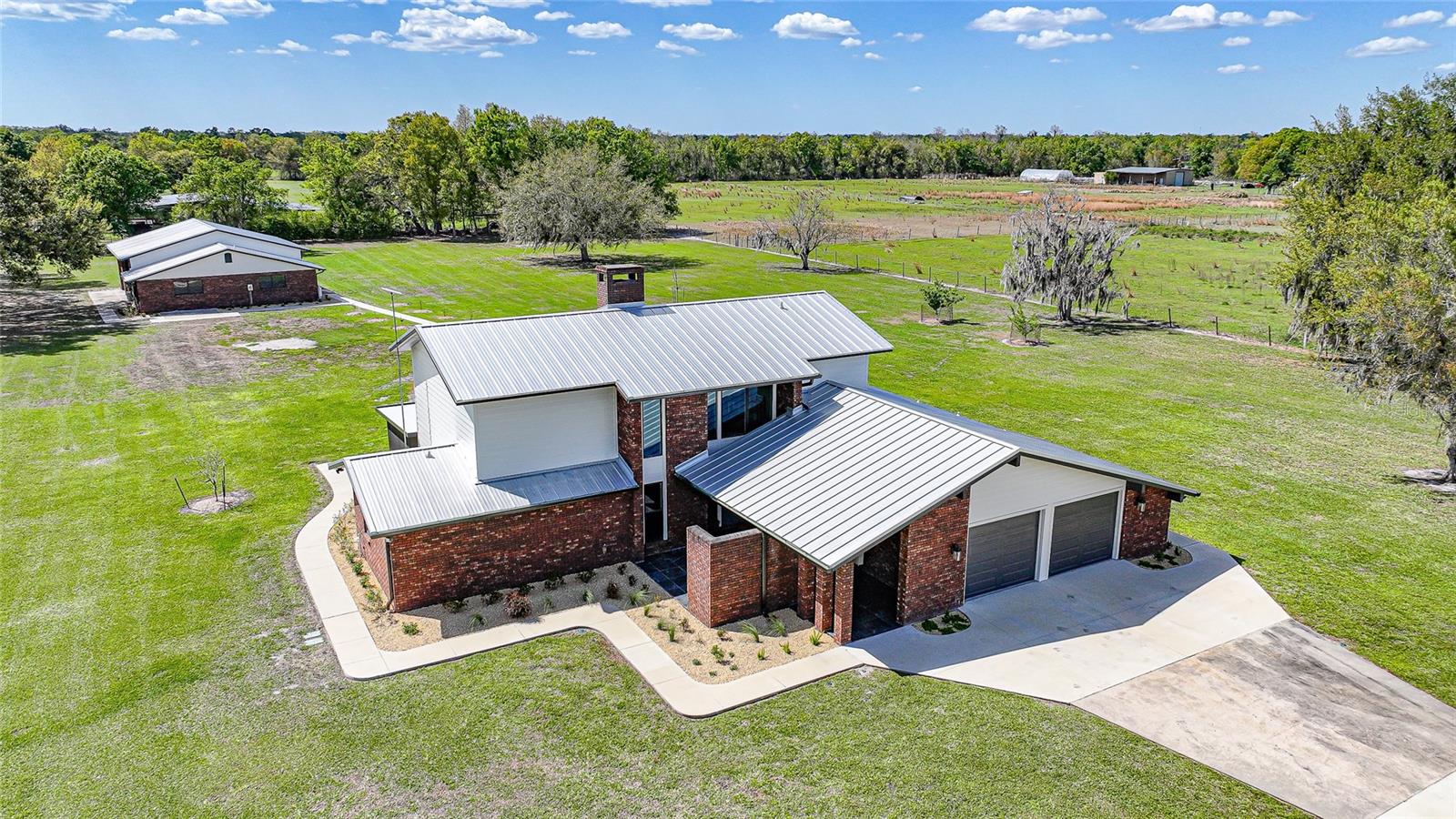 Welcome to your private 5 acre estate, where luxury meets functionality and every detail has been thoughtfully designed to impress. As you arrive, a long concrete driveway leads you to this stunning two-story brick home, showcasing exceptional curb appeal and a welcoming covered front porch. Step through the grand double doors into an impressive foyer featuring soaring ceilings and beautiful luxury vinyl flooring that flows throughout the main living areas. The heart of the home is the meticulously updated kitchen, perfectly suited for both everyday living and entertaining. It boasts quartz countertops, stainless steel appliances, pot filler, farmhouse sink, an oversized gas range with hood vent, and a convenient drawer microwave. The expansive double sided island offers ample seating, while the walk-in pantry is fully equipped with additional cabinetry, a second refrigerator, and freezer. Just off the kitchen, the dining area features a built-in coffee bar, creating a perfect space to start your day. The living room is equally impressive with its soaring ceilings, abundant natural light, and cozy wood burning fireplace. Each downstairs room offers direct access to the surrounding porches, allowing you to fully enjoy the peaceful views of your property. The spacious primary suite is located on the first floor and serves as a private retreat, complete with a large walk-in closet and a luxurious en suite bathroom featuring a standalone soaking tub and walk-in shower. The suite also includes its own private porch, perfect for relaxing in tranquility. Upstairs, you’ll find two generously sized guest bedrooms, each with oversized closets, connected by a walkway that overlooks both the living room and the grand entryway below. Additional interior features include a large laundry room and a versatile bonus space connected to the attached garage, offering endless possibilities for storage, a home office, or hobby area. Step outside and follow the beautifully landscaped concrete pathway past a serene koi pond with a filtration system, leading you to the impressive detached garage and potential in-law suite or man cave. The expansive 6+ car garage features five 11' foot garage doors and an open layout ideal for car enthusiasts, workshops, or a hobby farm setup, garage is accessible using the deeded side road/easement. Within the garage, the in-law suite offers two rooms, is fully air-conditioned, and includes rough-in plumbing for a future kitchenette and bathroom—ready for your customization. Built to stand out, this property also features a durable metal roof, hurricane impact windows, propane tankless water heater, gas dryer, wired for a generator and countless upgrades throughout. This one-of-a-kind estate offers space, luxury, and versatility all in one. Call today to schedule your private tour!