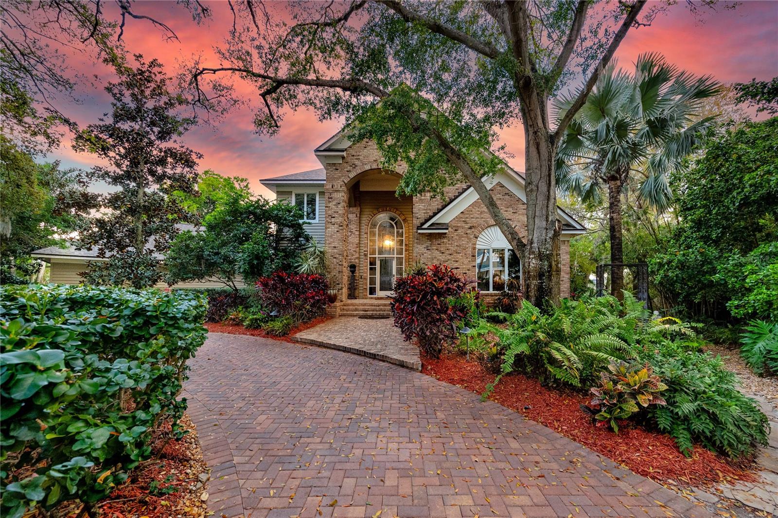 Golf View Estates is a quiet, cul-de-sac pocket of Palm Harbor where oversized lots back to conservation land. This property sits on three-quarters of an acre and grounds that function less like a backyard than a private botanical resort.
The outdoor entertaining complex is designed at a scale rarely found in residential Pinellas County. Terraced stone patios connected by illuminated paver walkways create multiple distinct zones: a covered wraparound lanai with travertine floors and ceiling fans across its full span, a custom outdoor kitchen with built-in stainless grill, warming drawers, prep sink, stone cabinetry, and granite counters, a fire pit terrace framed by curved stone retaining walls with recessed uplighting, and a substantial wood-beam pergola — large enough to host a live band — strung with Edison lights above its own dedicated gathering floor. The full complex comfortably accommodates 150 or more guests across its layered levels. Landscape lighting runs throughout: uplighting through the canopy of mature palms and tropicals, low path lights along every walkway, and ambient light embedded in the stonework that makes the grounds as compelling at night as they are at noon. The heated pool anchors the screened enclosure, centered on a hand-stacked stone waterfall grotto surrounded by tree ferns and bromeliads. A newer concrete seawall edges the conservation pond beyond. Flood insurance comes out to under $100 a month.
Inside, the home is 4,342 heated square feet across two floors, built of brick and ICF construction. The formal living and dining areas open the first floor with cathedral ceilings, crown molding, and rich wood doors with crystal and brass hardware throughout. Nearly every room orients toward the conservation and pool grounds outside. The kitchen is a considered split layout: solid wood cabinetry, granite countertops, a cooking island, dual breakfast bars, and a breakfast nook with a built-in granite-topped desk. A secondary dishwasher serves the wet bar, and a serving window connects directly to the screened lanai. The family room anchors the center of the home with a gas fireplace and custom built-in shelving, and five sets of French doors open to the wraparound rear deck, creating an indoor-outdoor flow that reads as intentional rather than incidental.
The first-floor primary suite is positioned well away from the main living areas, with pool and garden views, a solid wood built-out walk-in closet with an integrated organizational system, and a garden tub ensuite featuring split vanities, and a separate oversized walk in shower. A dedicated office with built-in closet sits adjacent on the first floor. Upstairs, two bedrooms each carry oversized walk-in closets — one with a window seat and dedicated makeup vanity — plus a full bath and an open loft currently used as a home gym, equally suited as a library or second living space. A three-car side-entry garage sits behind an oversized circular drive for privacy and accommodating guests.
Roof and HVAC replaced in 2023. Pool heater new in 2026. No HOA. Minutes from the Pinellas Trail, Dunedin's waterfront, and Honeymoon Island State Park, with both Tampa International and St. Pete-Clearwater Airport within approximately 30 minutes.