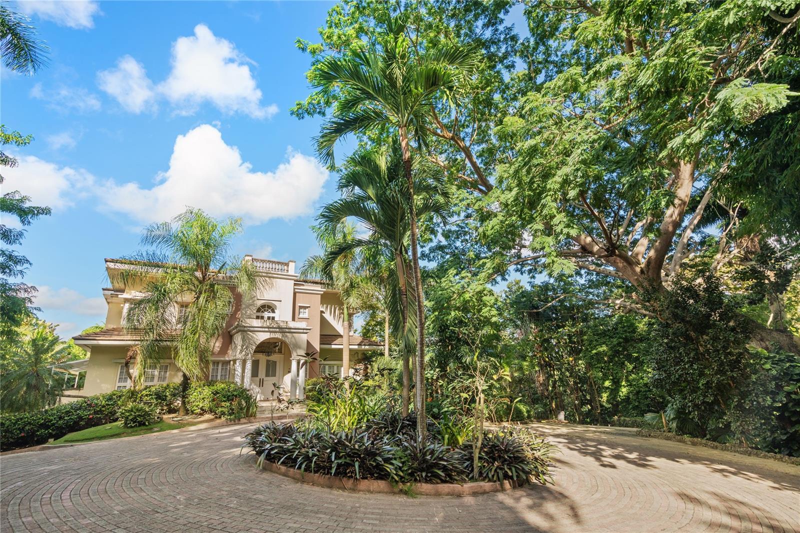 Located within the exclusive gated community of Tintillo Hills, this timeless custom-built residence offers privacy, scale, and refined living on an expansive 2.5-acre lot. The four-level estate features 11,415.24 sq ft of total construction, designed for both grand entertaining and comfortable full-time living.

The main level welcomes you with a grand foyer featuring soaring ceilings and a curved staircase, filled with natural light. This level includes formal living and dining rooms, a spacious kitchen with breakfast area, family room, and a versatile billiard room. Expansive open terraces and covered porches connect the interiors to the surrounding gardens and pool, creating seamless indoor-outdoor living.
The lower level offers exceptional amenities including a gym, sauna, wine cellar, full bathroom, maid’s room with en-suite bath, laundry area, extensive storage, and a three-car garage. Outdoor features include a pool, concrete terrace, manicured gardens, and a detached wood terrace with outdoor kitchen—ideal for entertaining.

The second floor features four bedrooms and a family room. The primary suite includes a spa-like bathroom, dual walk-in closets, and a private balcony overlooking the pool and gardens. Additional bedrooms offer full bathrooms and walk-in closets.
The third floor hosts a private office or library accessed by a spiral staircase, opening to a balcony with panoramic views extending to Old San Juan.

The estate is accessed through a private gated entrance with circular driveway and includes mature trees, expansive green areas, and a tennis court ready for renovation. The property is fully equipped with an industrial backup generator and a large-capacity water cistern, ensuring uninterrupted comfort and peace of mind. Adding a unique touch of countryside charm, the grounds also feature resident chickens, embodying the essence of campo en la ciudad—country living with city convenience.