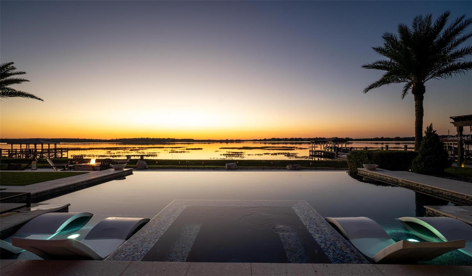 **A Rare Lakefront Peninsula Estate in Emerald Pointe**Ideally positioned at the tip of a private island peninsula, this stunning lakefront estate offers unmatched privacy and panoramic water vistas within **Emerald Pointe**, one of Winter Garden’s most exclusive double-gated enclaves. Set along the shimmering shores of **Johns Lake—ranked among Florida’s Top 10 Bass Fishing Lakes—**the residence captures uninterrupted lake views from the moment you step through the front door. Designed to blur the boundaries between indoors and out, the home features a **Double NanaWall system** that spans the main living space with floor-to-ceiling glass. When opened, the precision-engineered, dual-pane panels vanish completely, allowing the great room to merge seamlessly with the lakefront environment. When closed, the insulated glass provides exceptional sound and thermal protection, complemented by **motorized retractable screens** for effortless open-air living. This is a home created to live far beyond its walls. Offering **5 bedrooms, 4.5 bathrooms, a dedicated theater room, and a spacious loft**, the floorplan provides both luxury and livability. The **primary suite** enjoys its own expansive balcony overlooking the water—an ideal vantage point for the vivid, mirror-like sunsets that reflect across the lake each evening. A custom **100-bottle illuminated wine room** becomes a striking architectural feature, while meticulous craftsmanship is displayed throughout the home: rich wood beams, elegant crown molding, and bespoke built-ins surrounding a designer fireplace. The chef’s kitchen showcases a large center island finished with **Wolf and Sub-Zero appliances**, ideal for culinary enthusiasts and entertainers alike. Every room is **Sonos-ready**, and the home’s striking bar becomes the centerpiece of evening gatherings, complete with programmable lighting that syncs to music for a club-quality ambiance. Mechanical upgrades include **two new Amana variable-speed AC units** (3.5 & 4 ton), ensuring optimal efficiency and comfort year-round. Outside, resort living continues with an **infinity-edge pool** featuring a glass pebble finish, dual heaters, and bubblers, all framed by **Shellock resort-grade pavers** designed with natural seashell cooling technology. The **outdoor pergola summer kitchen** is an entertainer’s dream—complete with a grill, pizza oven, sink, refrigerator, app-controlled lighting, and Sonos-integrated audio. Waterfront amenities are second to none: a **140-foot Hodgskins-built dock** with a boat lift and two jet ski lifts provides instant access to the lake. The property also includes a **four-car garage** and a durable **tile roof**. From its world-class indoor-outdoor design to its truly unforgettable nightly sunset displays, this exceptional estate offers a lifestyle found nowhere else on Johns Lake. A private retreat—crafted for those who demand exceptional living at every turn.