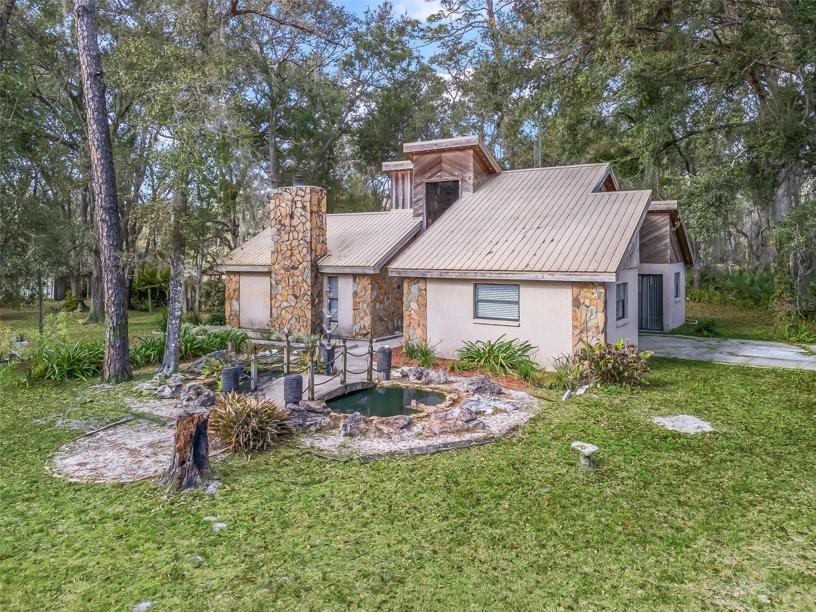 NEW PRICE on this tucked-away residence on 2.5+/- wooded acres, offering a rare blend of privacy, character, and creative potential. Surrounded by nature, the 2-bedroom, 2-bath block home is thoughtfully designed to blend seamlessly with its serene setting while providing a compelling foundation for refined customization. A dramatic spiral staircase serves as a sculptural focal point, ascending to a light-filled bonus retreat wrapped in rich cedar wood walls. An adaptable space well-suited for a home office, creative studio, entertaining lounge, or additional bedroom. Throughout the home, cedar wood ceilings add warmth and continuity, complemented by brand-new carpeting, while a striking stone fireplace anchors the main living space. Multiple sliding glass doors extend the interiors outward to patios and nature's tranquil views, creating seamless indoor-outdoor living and entertaining opportunities. The primary suite functions as a private sanctuary, complete with its own bath and direct patio access. The unfinished patio and elevated entertaining area present an exceptional opportunity to further enhance and personalize the outdoor living experience. The grounds reinforce the home’s retreat-like atmosphere, featuring a koi pond with a charming bridge, a fire pit, winding walking trails, including a concrete slab ideal for a future garage or workshop, along with a separate 10x20 storage building. Secluded, distinctive, and rich with potential, this one-of-a-kind property is ideal for those seeking a private woodland escape with the flexibility to craft a truly customized lifestyle. Perfectly balancing privacy with accessibility, it is conveniently located with easy access to I-75, U.S. Hwy 301, and State Road 50, offering a manageable commute to Ocala, Tampa, and Orlando. Just minutes from the Webster Flea Market, Sumter County Fairgrounds, and local shopping and dining. Outdoor recreation abounds with nearby lakes, nature trails, and the Withlacoochee State Forest. Enjoy the best of Florida living with Gulf Coast beaches and natural springs, approximately 45 minutes to one hour away. A rare offering where privacy, natural beauty, and architectural distinction converge is best appreciated through a private showing.