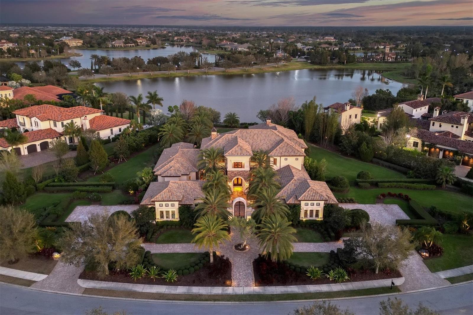 This incredible opulent estate sits on arguably one of the best lots in The Lake Club and is perfectly positioned on just over 1.5 acres to highlight the picturesque sweeping tranquil lake and clubhouse views. Anchorage sits behind another set of gates within the community. As you approach through the arched stone and iron gate you will be awed by the attention to detail on the brick and stone elevation. Built in 2012 by London Bay Homes, this home is impressive from the moment one enters the gated tree-lined walkway with meticulous landscaping and amazing oversized paver driveway. The remarkable archways and numerous details throughout this home are truly extraordinary. Exquisite designer lighting and draperies set this property apart from many others. This home features many living spaces both inside and out that flow together with pocketing sliding glass doors, travertine stone and wood flooring, with a cohesive color palette throughout. Just off the main living and dining room the walk-in climate-controlled wine room holds over 200 bottles and features a brick barrel ceiling and accent wall. The adjacent wet bar also features a brick barrel ceiling, upper and lower cabinetry, sink, and cabinet front beverage refrigerator. The gourmet kitchen is outfitted with custom wood cabinetry, stone tile backsplash, granite countertops, high-end stainless steel appliances, hidden walk-in pantry, desk area, an enormous functional island with counter height seating, and a large yet cozy eat-in area. The family room also has a wall of pocketing sliding doors that open to the covered patio creating one large entertaining space. The travertine floors extend to the patio and seamlessly expand the living space to take full advantage of the multiple covered and uncovered seating areas, wood ceilings, complete outdoor kitchen, large fire pit seating area, two-way stone fireplace, separate pool bath and enormous saltwater infinity edge pool with two fire bowls. The fenced-in backyard area is an absolute vision of paradise. The remote-controlled screens on the patio come down in between the stone columns to enclose most of the covered outdoor seating area. The luxurious owner’s suite is an intimate yet cozy space and features French door access to the patio, sitting area, two custom-organized walk-in closets, soaking tub, dual vanities, travertine walk-in shower all with a spa-like touch. The stunning stately office boasts a wood-beamed ceiling, brick archway, custom wood cabinetry, and direct access to the patio. As you walk up the wide wood staircase with a wrought iron railing you will find a generous in size bonus room equipped with theater seating for six, a projection screen, game room, and access to the covered terrace overlooking the pool, expansive water view and patio areas. There are 3 en-suite bedrooms for family or guests, as well as 2 gas fireplaces, an elevator, 2 laundry rooms, generator, impact windows, a bug misting system and plenty of storage closets throughout the home. There are two 2-car garages, one that is air conditioned and is currently being used as a home gym with travertine flooring. One of Lakewood Ranch’s premier guard gated communities, The Lake Club boasts a grande clubhouse with a restaurant, a bar area, two-resort style swimming pools, an expanded fitness center, massage and skincare room, a gaming and lifestyle center, tennis, pickleball, basketball, Linear Park and interlinking lakes for kayaking or fishing.