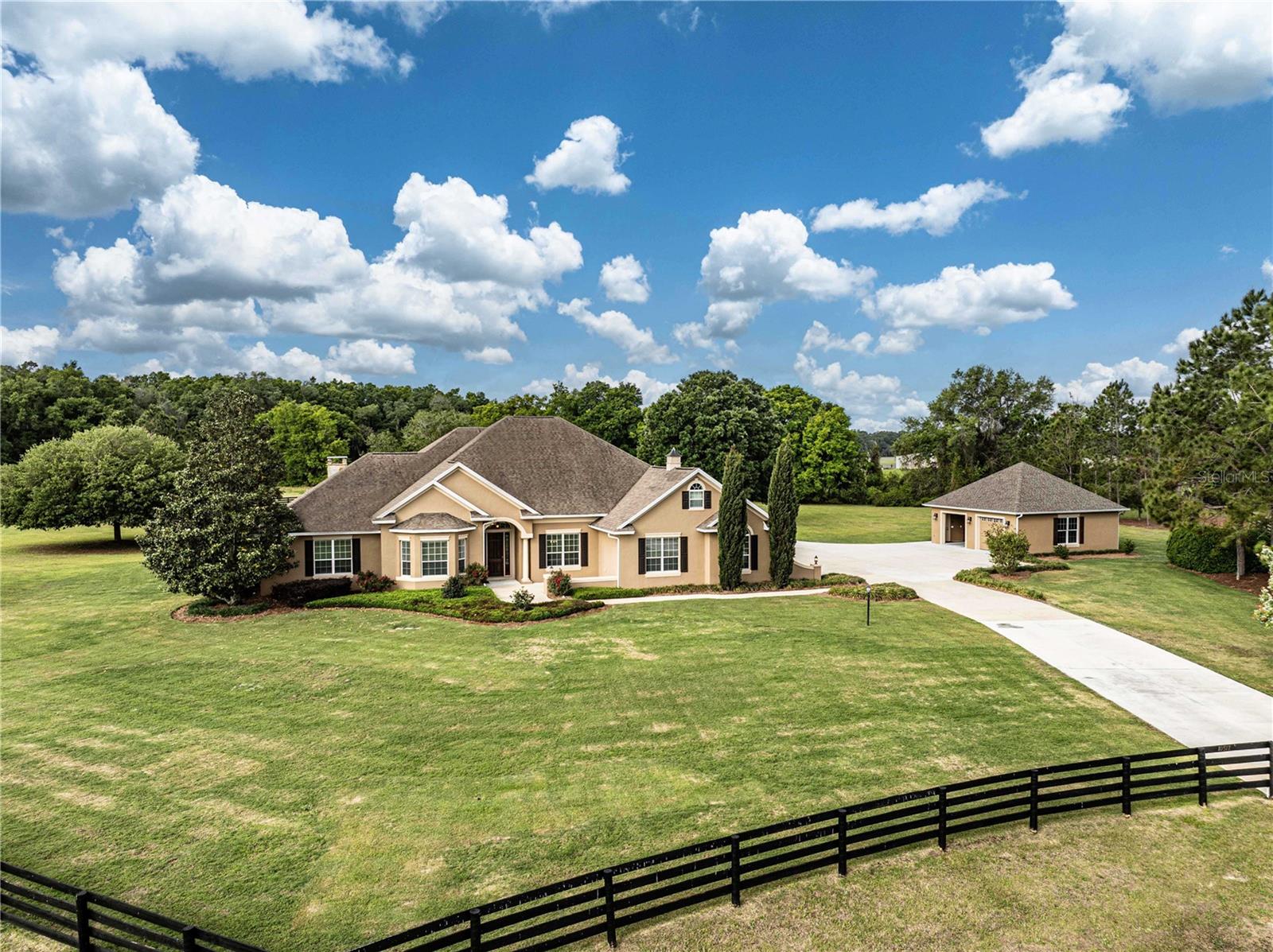 Welcome to a private luxury estate located in the gated community of Sumter Crossing in Oxford, offering timeless craftsmanship & resort-style living on 3.72 fully fenced acres just minutes from The Villages. This original owner, custom-built home, constructed in 2008 with solid block construction & a stucco exterior, features 3,705 square feet of heated & cooled living space (4,901 total square feet) with a thoughtfully designed split floor plan that blends elegance and functionality. Inside, you’ll find soaring tray ceilings, a formal dining room, & a versatile formal family room ideal for a piano, billiards, or additional entertaining space. The exquisite main living area showcases a custom accent wall  & opens seamlessly to the lanai through pocketing sliding glass doors that fully recess into the wall, creating true indoor-outdoor living. The chef’s kitchen is well equipped with an abundance of beautiful granite countertop space, including a serving and seating bar, stainless steel appliances, and an island prep area with pendant lighting. Additional features include a custom mudroom with built-in seating and storage, an interior laundry room with a deep sink and built-in ironing board, and a dedicated workout room complete with its own half bath. The owner’s suite serves as a private retreat w/ French doors leading directly to the pool area, a custom-built walk-in closet system, dual vanities, a garden tub, and a walk-in Roman shower. Guest accommodations include one bedroom w/ a private ensuite that also functions as a pool bath, along with a second guest bedroom & full hall bath. Step outside & experience what truly sets this property apart—an exceptional, resort-style pool & outdoor living space designed for both relaxation & entertaining, featuring a beautifully crafted stone pool with water feature, expansive screened lanai, outdoor kitchen with gas line for grilling, & a stunning floor-to-ceiling stone fireplace that anchors the space and creates an unforgettable setting year-round. Just beyond the lanai, enjoy a dedicated firepit area perfect for evenings under the stars, all surrounded by meticulously maintained landscaping and wide-open green space that enhances the privacy & estate feel of the property. The Bose surround sound system extends throughout both the interior & outdoor living areas, creating a seamless entertainment experience. The property also includes an attached two-car garage & a detached two-car garage, w/ the main garage featuring a custom electric fold-down stairway providing easy & safe access to a spacious, decked attic storage area complete w/ its own mini-split A/C system—ideal for climate-controlled storage. A whole-house water filtration system adds another level of comfort & quality to everyday living. Situated on 3.72 acres with four-board fencing in a gated, estate-style community, this property offers exceptional privacy while maintaining convenience w/ quick access to The Villages Charter Schools, Lake Sumter Landing & Brownwood Paddock Square, major roadways including I-75 and the Florida Turnpike, shopping such as Publix, Costco, & Walmart, a variety of dining options, and nearby medical facilities including UF Health. This is a rare opportunity to own a true estate property in one of Central Florida’s most desirable & rapidly growing areas—if you’re looking for privacy, luxury, & an unmatched outdoor living experience without sacrificing convenience, you’ve found your new home.