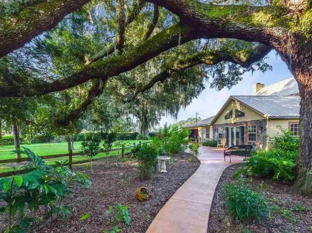 Set on 14 serene acres framed by Granddaddy oaks and open green space, this beautifully appointed country estate offers room to breathe, room to grow, and a lifestyle rooted in comfort and privacy. The 4-bedroom, 4-bath residence blends rustic character with modern functionality, featuring dual primary suites and thoughtful upgrades throughout.  Inside, clay tile flooring runs through the entire home—no carpet—paired with exposed wood beams, a knotty pine ceiling in the great room, and a wood-burning fireplace that brings warmth and charm to the dining area. The spacious open kitchen is designed for both everyday living and entertaining, showcasing quartz countertops, Shrock Amish cabinetry, stainless steel appliances, double ovens, an oversized island, and a generous walk-in pantry. A reverse-osmosis drinking system adds convenience.  The upstairs primary suite offers a private balcony overlooking the pool and a spa-style bath with a jetted corner tub and separate walk-in shower. A second primary suite downstairs includes its own soaking tub and walk-in shower, ideal for guests or multi-generational living. The expansive laundry room provides flexible space for crafts, hobbies, or recreation.  Outdoor living is a highlight, with a wide covered front porch overlooking the fenced acreage and fruit trees—pear, orange, lime, kumquat, and Meyer lemon. Composite stairs lead from the deck down to the pool area, creating a perfect spot to unwind. A whole-house generator (seller-owned propane tank) and a whole-house water filtration system installed in 2025 offer peace of mind.  A substantial barn on the property provides exceptional storage and utility options, featuring electric and plumbing, automatic watering, and oversized stalls that can be repurposed for equipment, vehicles, or future workshop space. An additional well and power pole in the pasture offer potential RV hookup capability.  A versatile outbuilding constructed in 2021–2022 adds even more flexibility, complete with concrete flooring, metal roof, running water with sink, on-demand water heater, window A/C unit, and both 110V and 220V electrical service. With minor modifications, this structure could serve as a workshop, studio, or future living quarters. Both the outbuilding and the home are wired for 110V and 220V, supporting a wide range of uses.  A rare opportunity to own a well-equipped acreage estate offering space, privacy, and the freedom to tailor the property to your lifestyle.