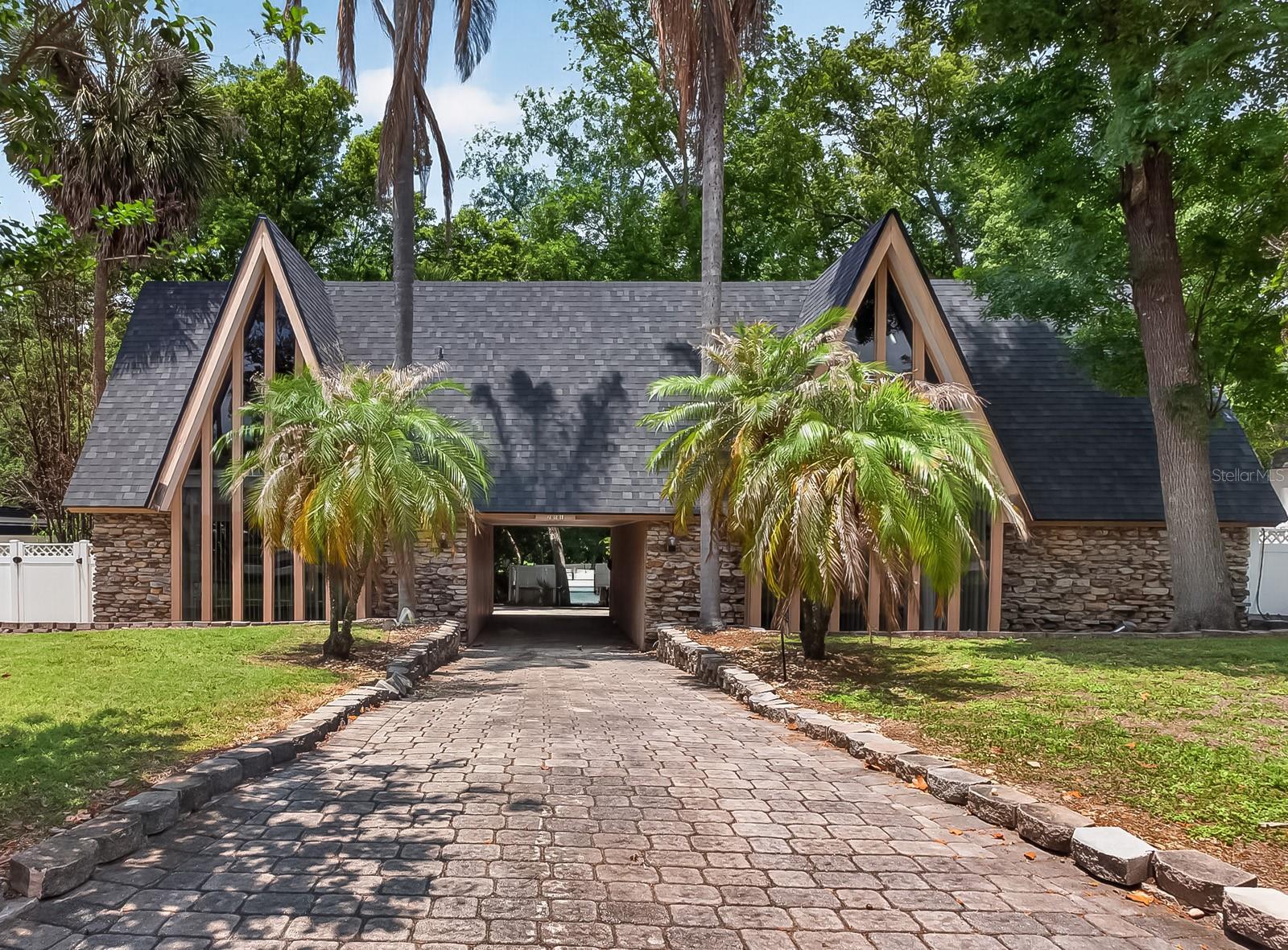 Located in one of Maitland’s established and tree-lined communities, this distinctive Mid-Century Inspired Chalet residence offers exceptional space, dramatic design elements, and endless potential for modern transformation. Positioned within sought-after school district  the home benefits from access to some of the area’s most well-regarded schools.
The home’s impressive pavered approach sets the stage for its chalet-inspired architecture, where soaring A-frame rooflines, dramatic window walls, and natural stone detailing create an unforgettable presence beneath a canopy of mature trees — where “Colorado ski chalet meets Old Florida.”
With 5 bedrooms and 3.5 bathrooms, the home is filled with high ceilings, expansive windows, and retro-inspired details that create character throughout. A striking open staircase with vintage styling showcases craftsmanship and design elements rarely found in today’s construction, while a massive paver courtyard welcomes guests and enhances the home’s unique indoor-outdoor lifestyle.
Inside, the formal dining and sunken living room feature coffered ceilings and a dramatic circular brick fireplace centerpiece, while walls of windows flood the interiors with natural light. The spacious kitchen includes a cooking island, double wall ovens, closet pantry, breakfast nook, and an abundance of windows overlooking the private backyard area.
Adding exceptional flexibility, the home also features a private in-law suite with its own separate entrance. The space includes a sleeping area, small seating/living space, private bathroom, kitchenette area with refrigerator and countertop workspace, and an eat-in area — ideal for multigenerational living, long-term guests, a private home office setup, or potential income-producing opportunities.
The upstairs bedrooms open onto a rear balcony, creating a true retreat feel among the mature trees and landscaped surroundings.
The primary bath features angular ceilings, oversized triangular windows, and a custom mosaic tile tub and double vanity that reflect the home’s original retro contemporary character.
Outdoors, enjoy a sparkling swimming pool surrounded by shade trees, an expansive paver deck, and a fully fenced yard ideal for entertaining, gardening, or creating a private oasis.
Recent updates include new roof 2024, along with a transferrable shingle warranty, both A/C HVAC units replaced 2019 and 2023, hot water heater was replaced in 2019, 2023 electrical work done, 2022 high end Samsung dishwasher installed, transferrable premium home appliance warranty expiring Feb 3 2027, gas-powered portable generator and underground utilities.  This home presents an exciting opportunity for buyers seeking architectural character and the chance to personalize a spacious residence with cosmetic updates including flooring, wall finishes, and modern design enhancements.
Conveniently located near top-rated dining, shopping, parks, and major commuter routes, Maitland remains one of Central Florida’s most desirable established communities, known for its timeless charm, mature landscapes, and exceptional location.