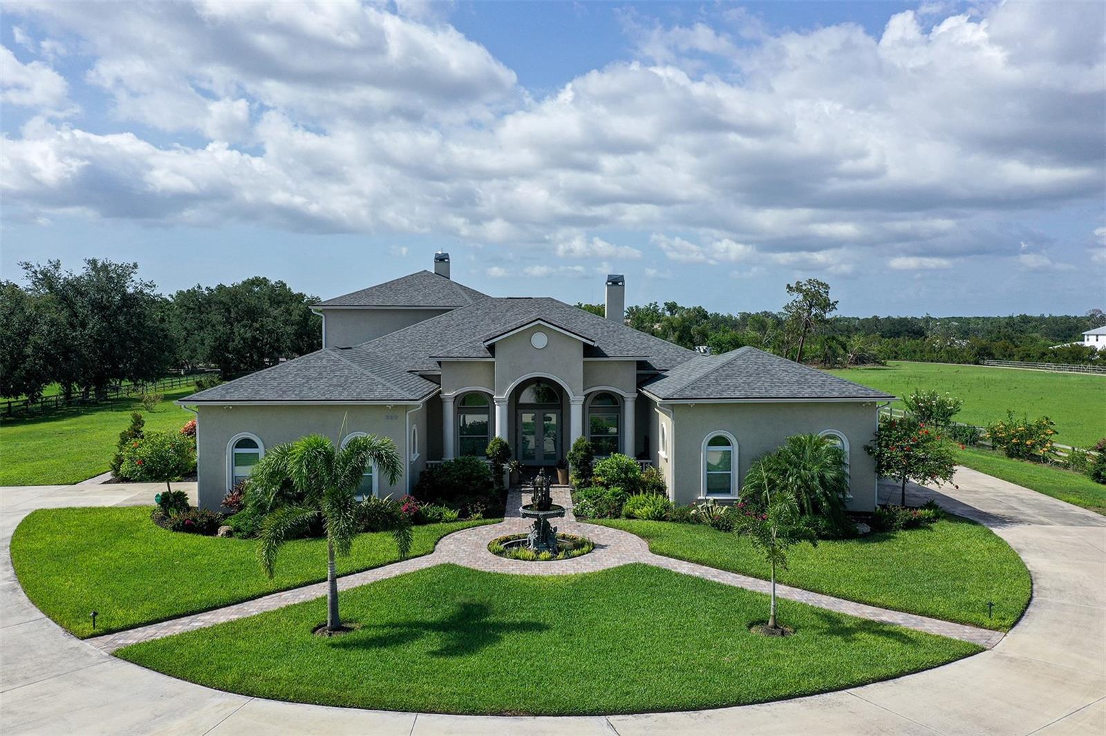 Quiet luxury, reimagined. Beyond a private electronic gate, a long, tree-lined drive winds past illuminated ponds and twin fountains, unveiling a stately 5,000+ sq ft estate set on over five serene acres. From the moment you arrive, this home feels like a private retreat—elegant, intentional, and impeccably updated. A brick-paved portico and welcoming courtyard with a custom bronze fountain set the tone as double French doors open to soaring 12-foot ceilings, tile floors, and a grand living room anchored by a fireplace. A private office with built-in shelving sits just off the entry, while the formal dining room impresses with its dramatic coved ceiling and French doors opening to a charming side patio. The heart of the home is a modern, beautifully appointed kitchen with stainless steel appliances, abundant cabinetry, and expansive counter space—seamlessly flowing into a sunny breakfast nook and inviting family room with a second fireplace. Nearby, a media room, tech closet with Wi-Fi tower, and half bath offer everyday convenience. Designed for flexibility and multigenerational living, the main level also features a private in-law suite with its own living/conference room, kitchenette, bedroom, full bath, and private entrance—currently used as a professional office suite. The luxurious primary retreat is also on the main floor, complete with two generous closets and a spa-like ensuite bath, along with a second ensuite bedroom, hobby room, and laundry room. Upstairs, a sweeping staircase leads to three additional bedrooms, two bathrooms, a cozy lounge area, a large storage room, and a covered balcony overlooking the grounds. Step outside to a screened-in pool and spa framed by stone pavers and generous lounging areas, all with peaceful views of the lush pasture beyond—perfect for entertaining or unwinding in complete privacy. In 2020, the entire property underwent a comprehensive renovation, including a new roof, plumbing, electrical wiring, multiple HVAC systems, new pool and spa, a number of upgraded windows, fresh landscaping and sod, brick pavers, concrete driveway, all new flooring, fully remodeled kitchen and baths, and multiple new fountains—giving the home an effective age of 2020. Additional upgrades include a 24-kW whole-house generator, reclaimed water irrigation across the full five acres, and a 500-gallon propane tank. This is a rare opportunity to own a thoughtfully renovated estate that delivers privacy, elegance, and exceptional value—all at an incredible price.
