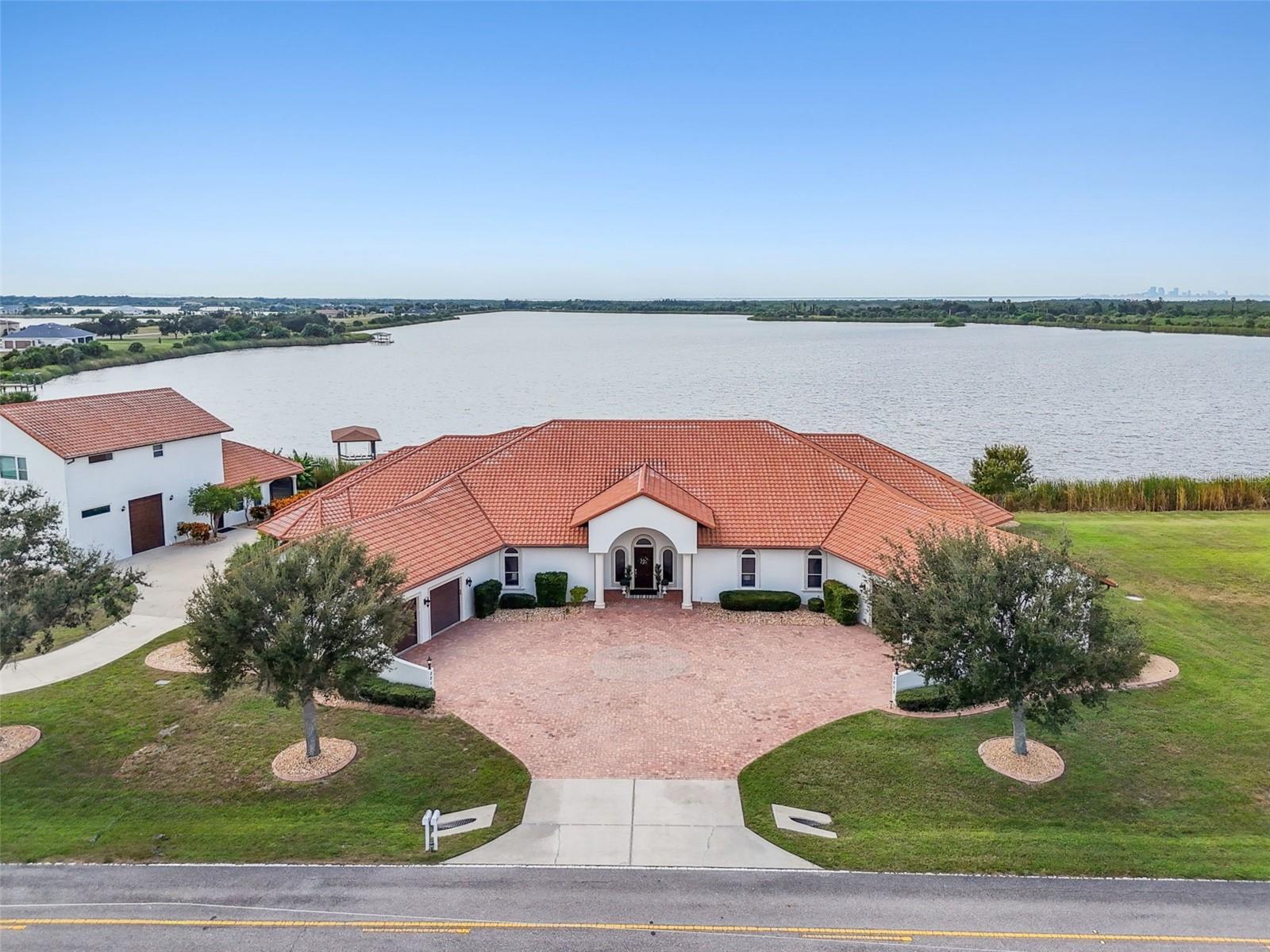 Tucked away within the exclusive enclave of Lost River Preserve, this custom-built Mediterranean-inspired waterfront estate offers a rare opportunity for multigenerational living without compromise. Newly repositioned to reflect exceptional value within Tampa Bay’s luxury market, this legacy property spans more than 2.25 acres across four addresses with over 300 feet of water frontage—thoughtfully designed to bring generations together while preserving connection and independence. From the moment you enter through the landscaped paver courtyard, the estate feels like a secluded waterfront sanctuary. Constructed with poured concrete tilt walls, impact-rated windows and doors, and a Galvalume aluminum tile-look roof, the home offers exceptional durability while positioned high and dry in Flood Zone X. Inside, nearly 6,700 square feet of refined living space unfolds across two distinct yet connected residences joined by a sunlit foyer—an ideal layout for extended family, aging parents, adult children, caregivers, or long-term guests. Soaring tray ceilings, arched architectural details, and walls of glass frame sweeping lake views throughout the home. The primary living areas and both primary suites were positioned to embrace the waterfront setting. Designed as a family compound, both primary suites feature spa-inspired baths, dual closets, and bay window seating overlooking the lake. Each residence offers its own office, ensuite bedrooms, generous storage, and dedicated laundry room. A flexible bonus suite with kitchenette and full bath opens directly to the pool terrace, ideal as a private suite, fitness studio, creative workspace, or independent living space. Outdoor living is equally impressive with a custom saltwater pool, 9-person spa, outdoor kitchen, and expansive screened lanai overlooking the water. The covered boat dock features a ski boat hoist and filleting station, while a second dock accommodates kayaks and paddleboards and is topped with a sunset bell. The detached villa offers approximately 1,200 square feet with a full kitchen and private accommodations, while the three-bay garage provides ample storage for boats and recreational equipment. An approximately 800-square-foot lakefront entertainment space with full bath and double French doors leading to a private screened lanai creates endless possibilities for gatherings, wellness experiences, family reunions, executive retreats, and extended-stay guests. Completing the estate is a fourth parcel pre-plumbed for a future residence, creating opportunities for expansion, guest accommodations, or a custom lakefront home. Set within the breathtaking 900-acre Lost River Preserve community, residents enjoy boating, trophy bass fishing, and an unmatched connection to nature surrounded by protected shoreline, scenic waterways, and native wildlife including roseate spoonbills, ospreys, and herons. Rich in history, the community is also home to the renowned Leisey Shell Pit fossil discovery, with artifacts showcased at the nearby Paleo Preserve Fossil Museum. Offering privacy without sacrificing convenience, this waterfront estate is located just 35 minutes from Downtown Tampa and within reach of fine dining, luxury shopping, championship sports, and world-class entertainment. Whether envisioned as a family compound, multigenerational estate, executive escape, or legacy property, this extraordinary waterfront residence delivers a lifestyle few properties can match!!