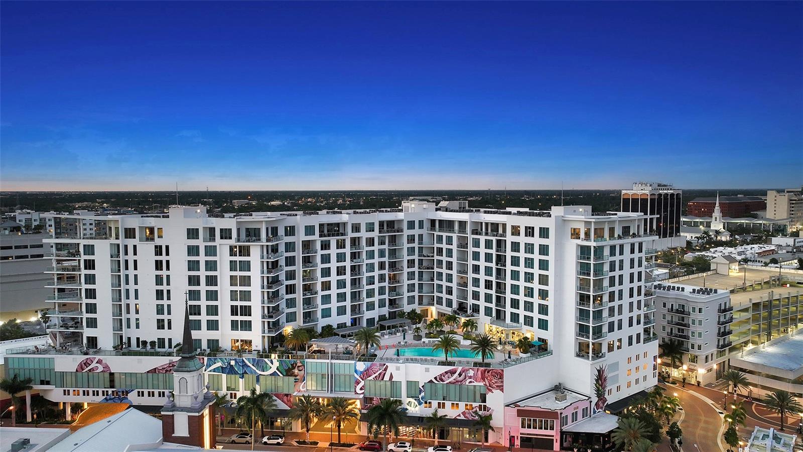 Residence 1013 at The Mark Sarasota offers a rare combination of comfort, convenience, and newer construction in the heart of downtown. Built in 2019, this residence provides modern peace of mind with impact hurricane glass and exterior doors, along with soaring 10-foot ceilings and a bright, open feel throughout. The home includes two deeded side-by-side covered parking spaces located just 20 feet from the elevator, an exceptional convenience rarely found downtown. Positioned on the quiet and highly desirable side of the building, the residence enjoys southern exposure and beautiful views overlooking the pool, with sightlines extending toward Sarasota Bay and the Gulf of Mexico. Set on the 10th floor, this thoughtfully designed layout features two bedrooms plus a den and two-and-a-half baths across 1,577 square feet, along with a covered terrace that extends the living space outdoors. The split-bedroom floor plan provides privacy while maintaining an open, functional flow ideal for both everyday living and entertaining. Walls of glass fill the home with natural light and frame tranquil views over the treetops toward Paul Thorpe Park and the Mermaid Fountain. The kitchen blends style and function with imported cabinetry, stone countertops, designer backsplash, stainless steel appliances, and an upgraded induction cooktop. It connects seamlessly to the main living and dining areas, creating an inviting space to gather. A full-size utility room with additional storage adds everyday practicality.  The primary suite opens directly to the terrace and features a spacious walk-in closet and a well-appointed bath with illuminated mirrors and refined finishes. The guest bedroom is privately situated, while the den offers flexibility as a home office or additional lounge space. A climate-controlled storage unit located just down the hall provides added convenience. Residents enjoy The Mark’s resort-style amenities, including a heated saltwater pool and spa, outdoor seating and grilling areas, club room with catering kitchen, billiards, fitness center, yoga studio, and spin room. The building also offers a pet-friendly environment, 24/7 front desk staffing, secure lobby access, and overnight roving security. Located in the center of downtown Sarasota, the residence places you just steps from the renowned Sarasota Farmers Market and only two blocks from Whole Foods Market. Cultural attractions including the Sarasota Opera House, Florida Studio Theatre, and Selby Public Library are all within easy reach, along with dining, galleries, and the waterfront. Surrounded by exciting new luxury developments, including the nearby Waldorf Astoria Residences Sarasota, this location continues to elevate as one of downtown’s most sought-after areas. Don’t miss the opportunity to own a newer-built residence in one of Sarasota’s most vibrant and walkable neighborhoods.