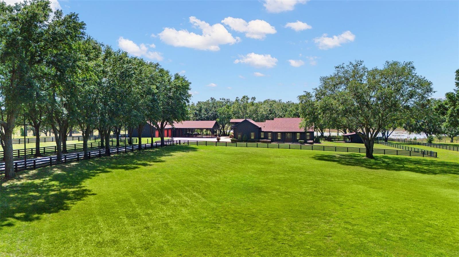 Situated on 5 pristine acres, this exceptional lakefront property offers a rare blend of privacy, space, and natural beauty. The fully fenced grounds feature classic black wood fencing, a picturesque oak-lined driveway with elegant lighting, manicured landscaping, mature trees, and expansive green space—all framed by peaceful water views.
The thoughtfully designed residence showcases timeless materials and warm architectural details throughout. Inside, you’ll find three spacious bedrooms with rich wood flooring, two full bathrooms, and a half bath. The open-concept great room seamlessly connects the kitchen, dining, and living areas, highlighted by custom wood cabinetry, tile flooring, wood ceilings, and built-in entertainment features. A flexible office or gym with Chicago brick and wood flooring adds versatility, while a second-floor bonus room offers additional space for a media room, playroom, or private retreat. The exterior is equally impressive with black-painted James Hardie siding and Chicago brick front and rear patios with wood ceilings, creating inviting outdoor living spaces.
The property functions as a true private compound with exceptional outbuildings. A detached lakefront entertainment retreat captures stunning sunset views and includes a kitchen, full bath, dining area with convertible poker table, and ample storage—perfect for hosting or relaxing.
Car enthusiasts will appreciate the custom climate controlled garage and workshop featuring 10’ entry doors, multiple vehicle lifts, epoxy flooring, storage systems, and elevator access to a second-level loft lounge. An architecturally designed carport accommodates up to six vehicles and doubles as a covered pavilion for gatherings, complemented by an aluminum pergola and a Colorado stone propane fire boulder for outdoor entertaining. A separate outbuilding offers flexible use as a playhouse, pet space, hobby room, or studio.
The property is equipped with a comprehensive security system including surveillance cameras, driveway alerts, and emergency strobe notifications. A Cummins 30kW generator powers the home and all outbuildings, ensuring reliable backup for the entire estate.
Located just minutes from Hamlin Town Center, enjoy convenient access to top dining, shopping, entertainment, and everyday essentials, along with quick connectivity to SR-429, Walt Disney World, Downtown Winter Garden, and the greater Orlando area—offering the perfect balance of accessibility and serene living.