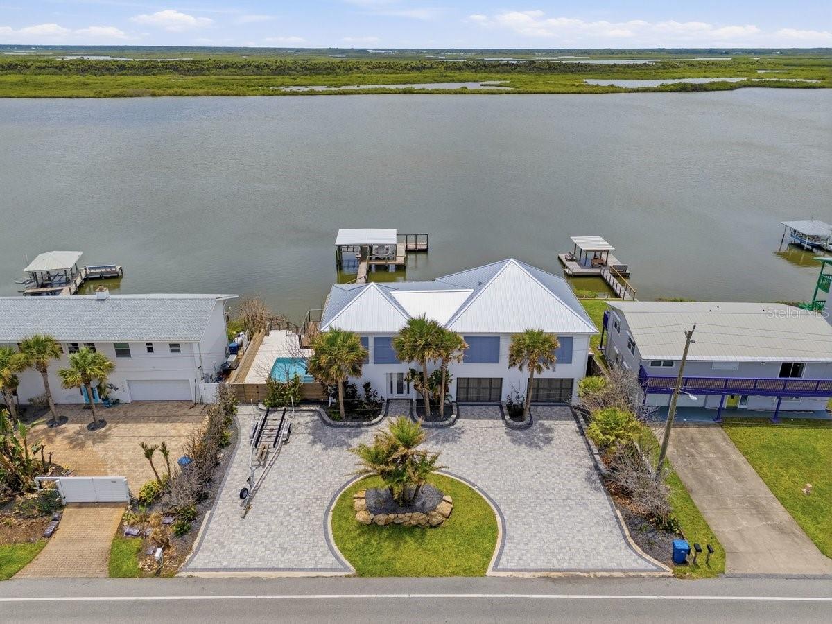 Nestled on 100 feet of pristine Intracoastal Waterway frontage in the coveted Bethune Beach neighborhood of New Smyrna Beach, this extraordinary custom-built coastal estate delivers an unrivaled blend of luxury, craftsmanship, and waterfront lifestyle just steps from a private beach walkover and the untouched beauty of Canaveral National Seashore. Completed less than 3 years ago with no expense spared, this fully furnished turnkey masterpiece showcases breathtaking panoramic river views with no homes across the water, offering unmatched privacy and spectacular sunsets. Elevated high above the water with a seawall, multi-tier retaining walls, and lush tropical landscaping, the property features an IPE dock complete with water, cleaning station, floating dock, and 2 covered boat lifts. A circular paver driveway accommodates up to 10 vehicles in addition to the oversized 36' wide two-car air-conditioned garage featuring tiled floors, 10' ceilings, custom garage doors with side-mount openers, wet bar with Hoshizaki commercial ice machine, Thermador wine cooler, 2nd laundry, and massive storage room for all your gear. Inside, dramatic architecture and refined modern design immediately captivate with smooth walls, a stunning underlit floating IPE staircase with cable rails, and a commercial-grade elevator with auto-glass gate offering unforgettable water views as it ascends to the main living level. The first floor hosts 2 luxurious riverfront guest suites and direct access to the resort-style pool with travertine decking and covered lanai. Upstairs, soaring vaulted ceilings frame the expansive riverfront living, dining, and entertaining spaces, all opening through walls of impact sliders to covered and open-air IPE-clad terraces with electric sunshades overlooking the water. The showstopping chef’s kitchen is truly one of a kind, appointed with Sub-Zero refrigeration and freezer drawers, Viking gas range with custom hood, double Thermador wall ovens, Bosch microwave drawer, prep sink, quartz countertops, and wine cooler. The lavish primary suite offers double French door entry, endless water views, custom walk-in closet, and an extraordinary spa-inspired bath featuring Brizo rain heads and body jets controlled by Moen digital panels, quartz vanities, illuminated mirrors, soaking tub, and smart Brondell toilet. A massive second-floor laundry room with dual access from both the primary suite and kitchen adds exceptional everyday conveniencen. Quarter-sawn oak flooring spans the second level while oversized designer tile flows throughout the lower level and baths. Additional luxury features include aluminum metal roofing, hurricane impact windows and doors, concrete beam-and-column construction with reinforced concrete floors and masonry exterior, Aquasana whole-home filtration, hybrid water heater, Honeywell security system with cameras, recessed LED lighting, electric blinds for west-facing windows, Moen digital shower systems, smart toilets throughout, built-in wifi expanders, surround sound, and premium Arhaus furnishings included, in addition to Polywood exterior seating. Located just minutes from groceries, fine and casual dining, shopping, and vibrant nightly entertainment, this is a rare opportunity to own one of Bethune Beach’s most exceptional modern waterfront estates. ALL INFORMATION IS ASSUMED ACCURATE BUT NOT GUARANTEED. ALL MEASUREMENTS ARE APPROXIMATE. BUYERS AND THEIR AGENTS SHALL VERIFY INFORMATION DEEMED IMPORTANT TO THEM.