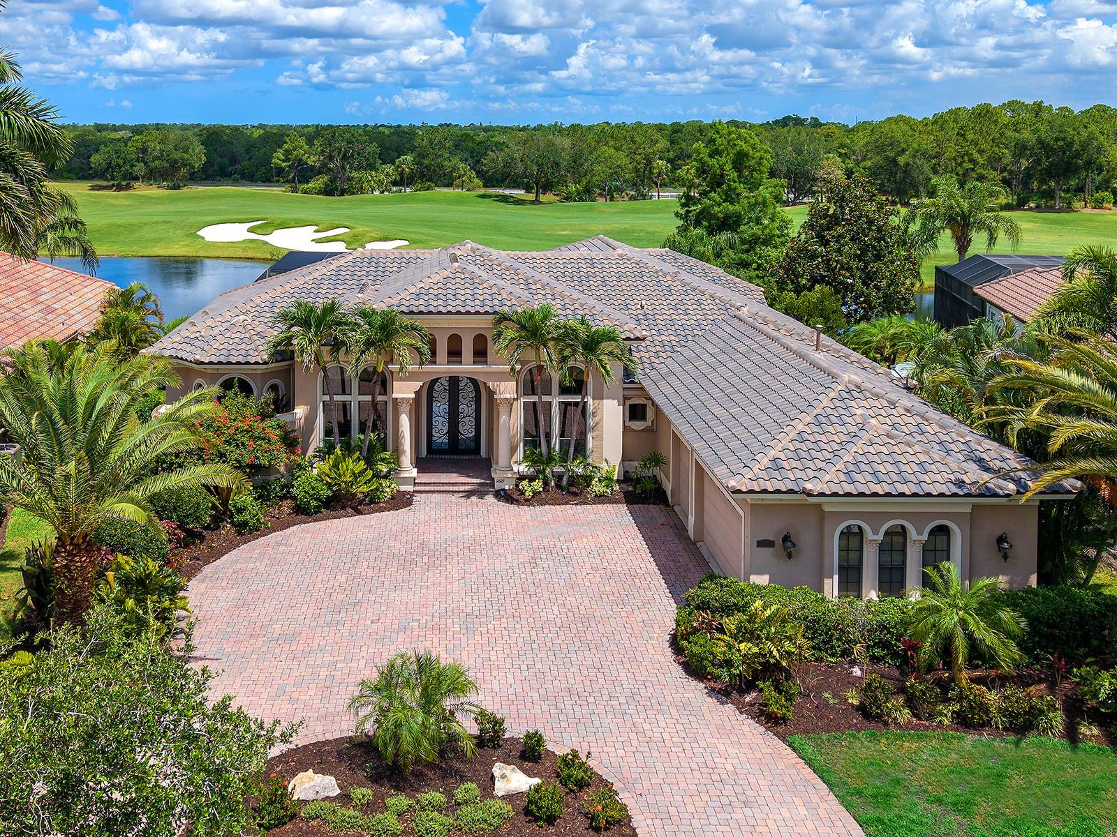 Positioned along the 14th fairway of the Kings Dunes Golf Course within the gated Lakewood Ranch Country Club, this custom John Cannon residence captures tranquil golf course and water views while offering timeless Mediterranean-inspired architecture and exceptional indoor-outdoor living. Equipped with a new roof, hurricane shutters for the windows and doors, a whole-home Generac generator, and a tankless water heater, the residence blends timeless design with thoughtful storm preparedness and peace of mind. Situated on a private 0.33-acre homesite, the property features 4 bedrooms, 3 full baths, 1 half bath and 3,887 square feet under air. A circular brick paver driveway, decorative cast stone columns, mature tropical landscaping and arched architectural detailing create striking curb appeal with estate-style presence. Wrought-iron and glass double entry doors open to interiors defined by soaring ceilings, layered crown molding, custom millwork, recessed lighting, architectural niches, polished travertine flooring and expansive picture windows framing the pool, water and golf course views beyond. Designed for both everyday living and entertaining, the living and dining rooms are filled with natural light and enhanced by tray ceilings, custom trim detailing, decorative ceiling accents and walls of windows. The family room features a custom-built-in media wall and pocketing sliding glass doors that create a seamless connection to the outdoor living spaces and screened lanai. The open-concept kitchen blends style and functionality with custom espresso cabinetry, illuminated glass-front display cabinets, granite countertops, full-height stone backsplash, center island with prep sink, walk-in pantry and premium Monogram stainless steel appliances, including a built-in wall oven and microwave combination, warming drawer, wine chiller and six-burner gas cooktop with grill insert. A sunlit breakfast nook with a mitered picture window and French doors overlooks the pool, lake and fairway views. The private library/office serves as a standout feature of the home, complete with floor-to-ceiling built-in shelving, integrated rolling ladder systems, rich wood flooring and expansive transom windows. The primary suite offers direct lanai access, panoramic pool and golf course views, dual walk-in closets with built-in organization systems and a spacious en-suite bath featuring hammered copper sinks, soaking tub, new carpet, dual vanities and a walk-in shower overlooking a private outdoor atrium. Three additional guest bedrooms provide comfortable accommodations for family and visitors, while thoughtfully designed guest baths showcase designer wallpaper, mosaic tile accents, custom cabinetry and glass-enclosed showers. Outdoor living is designed for year-round enjoyment with an expansive screened lanai just recently completely restored and re-screened, resort-style heated pool and raised spa with spillover water feature, brick paver pool deck, outdoor fireplace and a custom summer kitchen with built-in Monogram grill, prep sink and bar seating. Multiple covered lounging and dining areas overlook the peaceful water and fairway setting, creating an ideal environment for entertaining or quiet relaxation. Completing the property is a side-entry three-car garage with smooth-finish flooring and overhead storage potential, all within one of Lakewood Ranch’s premier golf course communities.