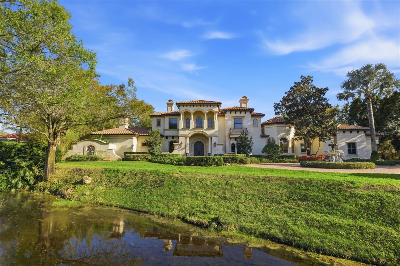 Located within the gated Steeplechase community of Odessa, this lakefront estate spans more than three private acres and offers 6,749 square feet of thoughtfully designed living space. A tree-lined paver drive leads to a circular driveway with fountain and a stately façade featuring arched windows and manicured landscaping. The property provides direct lake frontage and a high level of privacy within one of Tampa Bay’s most established luxury neighborhoods. The residence includes five en-suite bedrooms and six full baths, with a primary suite on the main level featuring a fireplace, flex space, and spa bath. Interior spaces are defined by soaring ceilings, marble floors, formal living and dining rooms, and a chef’s kitchen equipped with Wolf and Sub-Zero appliances. Additional rooms include a wood-paneled office, climate-controlled wine cellar, and dedicated lounge, offering flexibility for work, entertaining, and private use. Outdoor living is arranged across multiple covered and open-air areas, including a columned lanai, pool and spa, and a fully equipped summer kitchen overlooking the lake. Notable features include a four-car garage with dual EV chargers, central vacuum system, dog-washing station, and whole-house generator. A well-appointed waterfront property combining acreage, security, and proximity to Tampa Bay amenities.